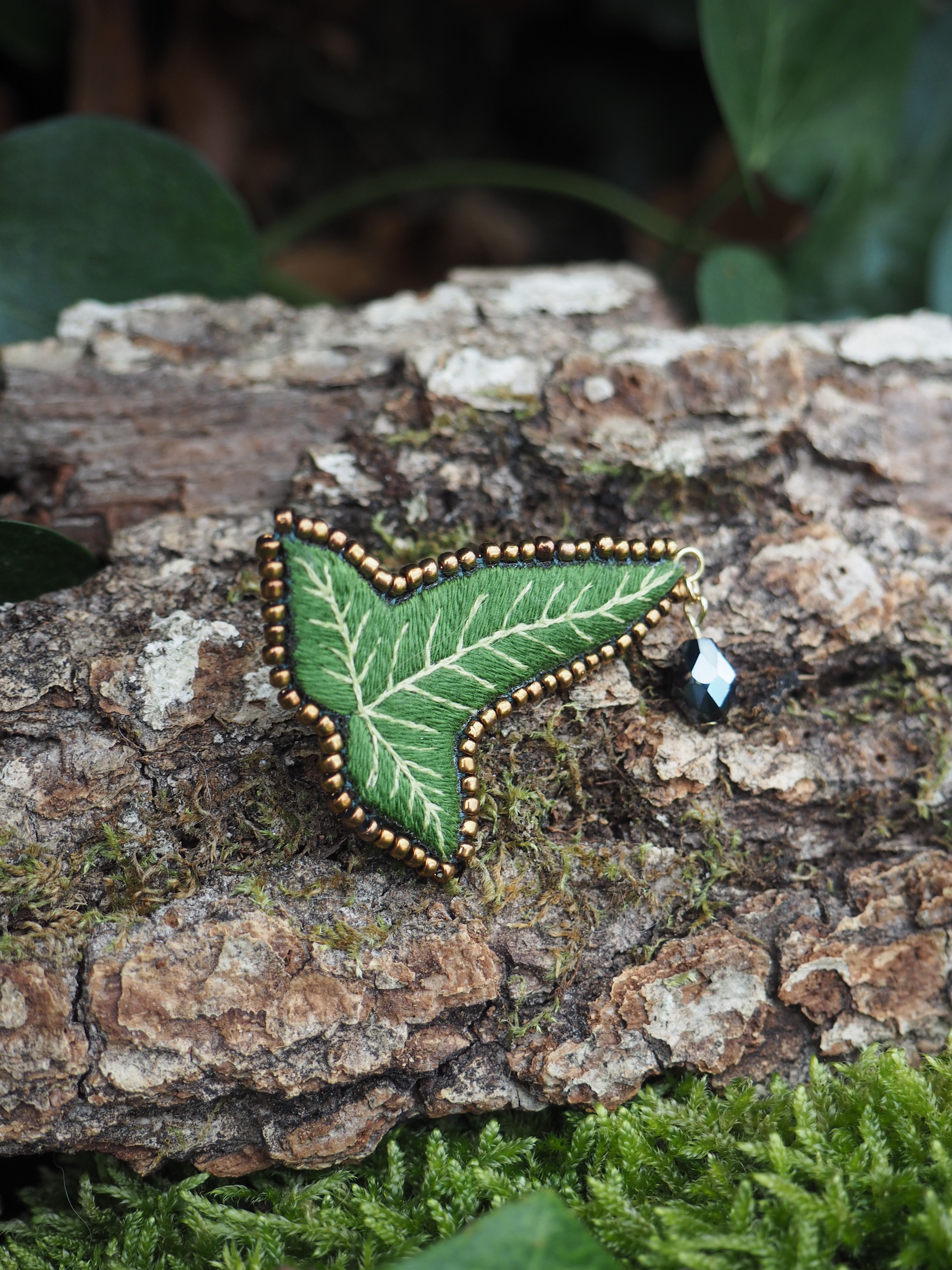 Cette image présente une broche en forme de feuille de lierre, posée sur un morceau de bois recouvert de mousse. Voici une description détaillée des éléments visuels : *   **La broche :** C'est l'élément central de l'image. Elle est conçue pour ressembler à une feuille de lierre, avec une forme lobée caractéristique.
    *   **Couleur et texture :** La feuille est d'un vert vif, avec des nervures blanches brodées qui imitent l'aspect naturel d'une feuille. Le contour de la feuille est orné de petites perles dorées, ajoutant une touche d'éclat et de finition.
    *   **Détail supplémentaire :** Accroché à l'une des pointes de la feuille, il y a un petit pendentif ou une perle de couleur sombre (peut-être noire ou bleu foncé) avec une facette brillante, qui capte la lumière.
*   **Le support :** La broche est posée sur un morceau de bois mort, probablement une branche ou un tronc.
    *   **Texture du bois :** Le bois a une écorce rugueuse et texturée, avec des crevasses et des zones plus lisses. Sa couleur est un mélange de bruns et de gris.
    *   **Mousse :** Une fine couche de mousse verte, de couleur plus foncée que la broche, recouvre certaines parties du bois, notamment sur le côté inférieur droit et en arrière-plan.
*   **L'arrière-plan :** L'arrière-plan est flou, ce qui met en valeur la broche. On distingue des formes vertes indistinctes, suggérant d'autres plantes ou feuillages, renforçant l'ambiance naturelle.
*   **Lumière et ambiance :** La lumière semble naturelle et douce, éclairant bien la broche et faisant ressortir ses détails. L'ensemble de l'image dégage une atmosphère calme et naturelle, mettant en valeur l'artisanat de la broche. En résumé, l'image est une photographie macro d'une broche artisanale en forme de feuille de lierre, mise en scène dans un environnement naturel pour souligner son inspiration botanique.
