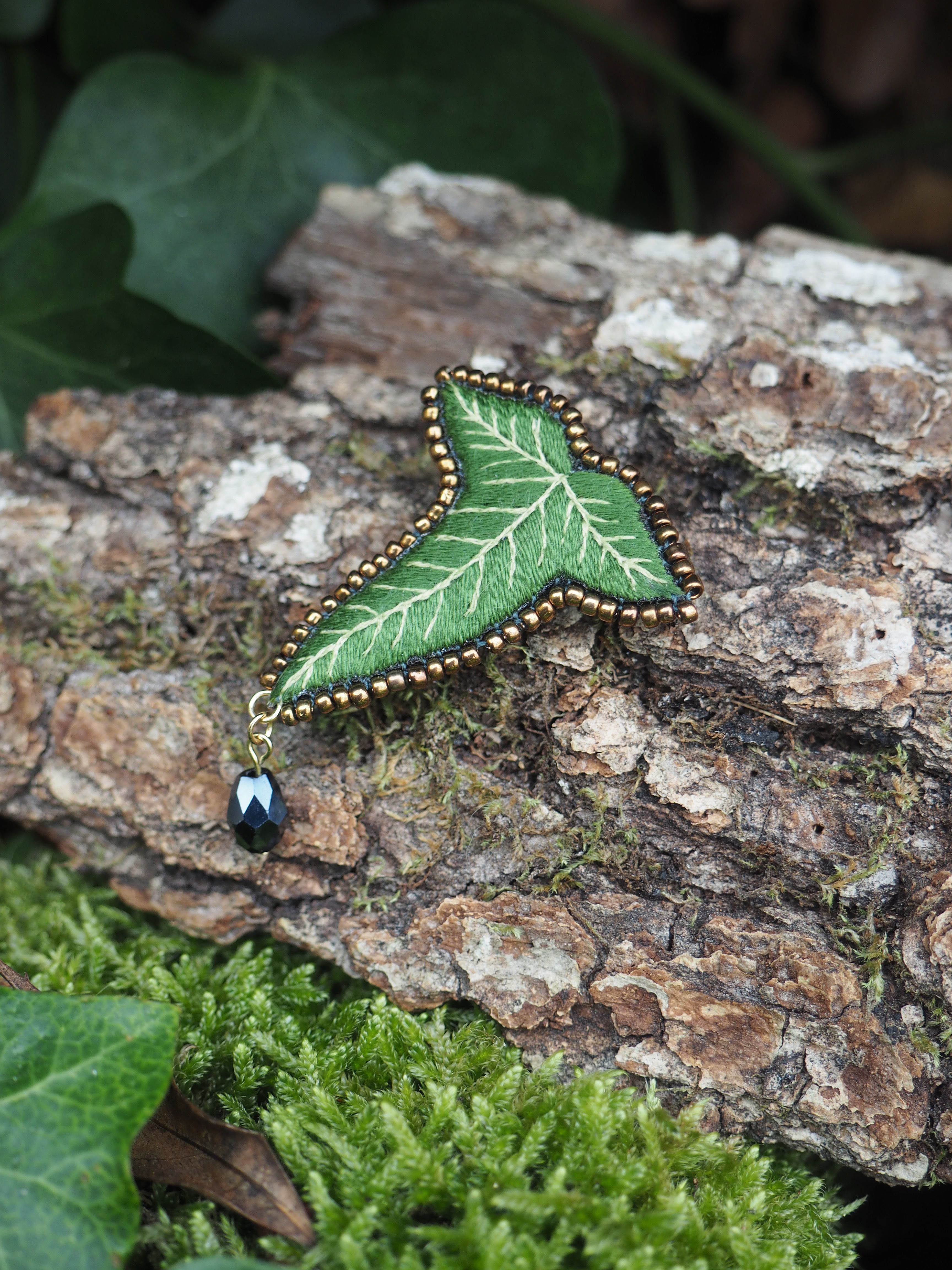 L'image présente une broche en forme de feuille de lierre, posée sur un morceau d'écorce d'arbre texturée et recouverte de mousse verte. La broche est le point focal de l'image. Elle est de couleur verte, avec des nervures plus claires brodées qui imitent celles d'une vraie feuille de lierre. Le contour de la broche est orné de petites perles dorées, ajoutant une touche d'éclat. Une petite perle noire brillante est suspendue à la pointe inférieure de la broche par un anneau doré, agissant comme un petit pendentif. L'arrière-plan est composé d'éléments naturels. Le morceau d'écorce sur lequel repose la broche est rugueux et présente des nuances de brun et de gris, avec des taches de mousse verte plus foncée. En dessous et autour de l'écorce, on peut voir une couche dense de mousse verte vive, créant un contraste de texture et de couleur. Quelques feuilles de lierre naturelles, de couleur vert foncé, sont visibles dans le coin inférieur gauche et en haut à gauche de l'image, renforçant le thème naturel et végétal. La lumière semble naturelle et douce, mettant en valeur les détails de la broche et les textures de l'environnement. L'ensemble de l'image dégage une atmosphère naturelle et délicate, soulignant le caractère artisanal et la beauté de la broche.