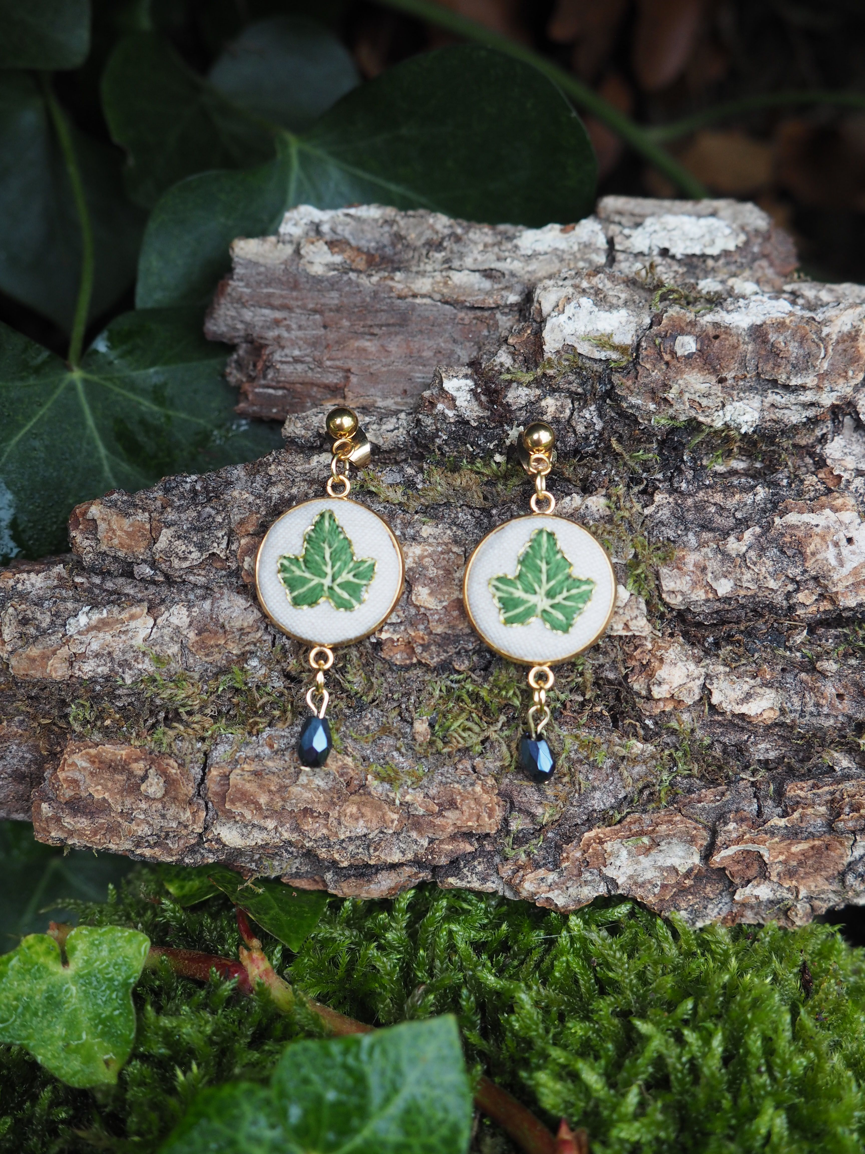 This close-up shot features a pair of nature-inspired earrings resting on a piece of tree bark, surrounded by lush green foliage. The earrings are identical, each consisting of a gold-colored stud from which an oval-shaped pendant hangs. The pendant has a gold-colored rim and a white background, upon which a detailed green leaf design is depicted. Below the oval pendant, a small, dark, teardrop-shaped bead dangles, adding a subtle touch of elegance. The tree bark beneath the earrings is textured, with visible grooves and patches of green moss, suggesting an outdoor or natural setting. In the foreground and background, various green leaves and moss create a soft, organic backdrop, enhancing the natural theme of the jewelry. The lighting appears soft, highlighting the details of the earrings and their surroundings.