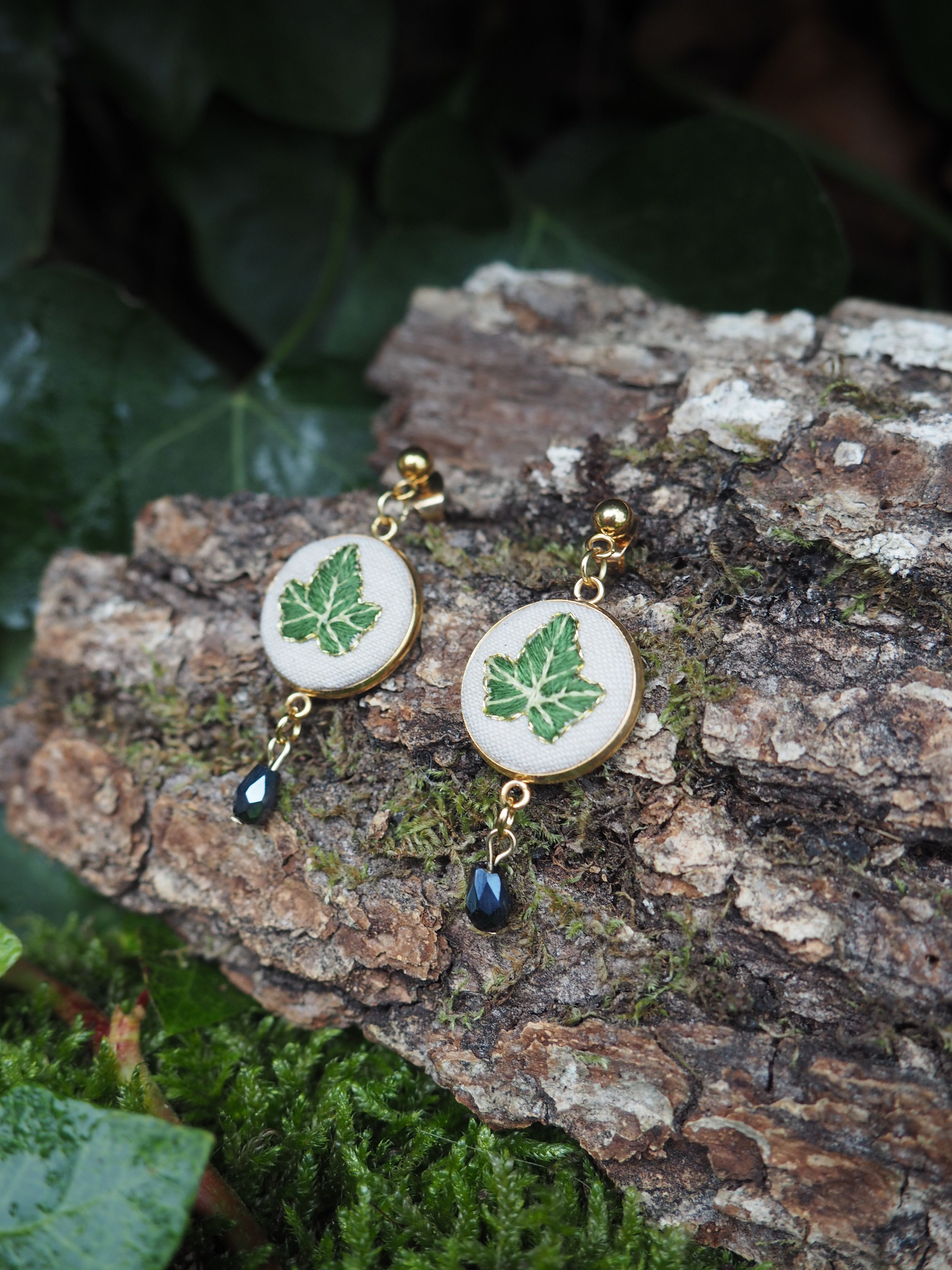This image features a pair of handcrafted earrings, each adorned with a delicate green leaf design, set against a natural, rustic backdrop. Here's a breakdown of the visual properties: **Subject:**
*   **Earrings:** There are two earrings, appearing to be a matching pair.
    *   **Design:** Each earring consists of a circular, flat component with a white or off-white background. Centered on this background is a detailed green leaf, possibly an ivy leaf, with visible veins. The leaf design has a slightly raised or embroidered appearance.
    *   **Frame:** The circular component is framed by a thin, golden-colored metal bezel.
    *   **Dangle:** Attached to the bottom of each circular component is a small, dark, faceted bead (possibly glass or crystal) that dangles.
    *   **Hook/Stud:** At the top of each earring, there's a small, golden-colored ball, suggesting they are stud earrings or have a stud-like attachment.
    *   **Material:** The golden elements appear to be metal, possibly brass or gold-plated. The white background could be fabric, resin, or enamel. The green leaf looks like a miniature embroidery or a detailed print under a clear coating. **Setting/Background:**
*   **Natural Elements:** The earrings are displayed on a piece of rough, textured tree bark, which is covered in patches of vibrant green moss.
*   **Foliage:** In the background, out of focus, are dark green leaves, likely ivy, contributing to a natural, woodland aesthetic.
*   **Color Palette:** The dominant colors are greens (moss, leaves, leaf design on earrings), browns (bark), and gold (earring metal), with touches of white/cream and dark blue/black from the dangling beads. **Composition and Lighting:**
*   **Arrangement:** The earrings are placed side-by-side on the bark, slightly angled, allowing both to be clearly visible.
*   **Depth of Field:** The foreground (earrings and bark) is in sharp focus, while the background foliage is softly blurred, creating a sense of depth and drawing attention to the jewelry.
*   **Lighting:** The lighting appears natural and soft, possibly overcast or diffused, highlighting the details of the earrings without harsh shadows. **Overall Impression:**
The image conveys a sense of natural beauty, craftsmanship, and elegance. The choice of a natural setting complements the leaf motif of the earrings, suggesting an organic or botanical theme. The focus is clearly on showcasing the intricate details of the handmade jewelry.