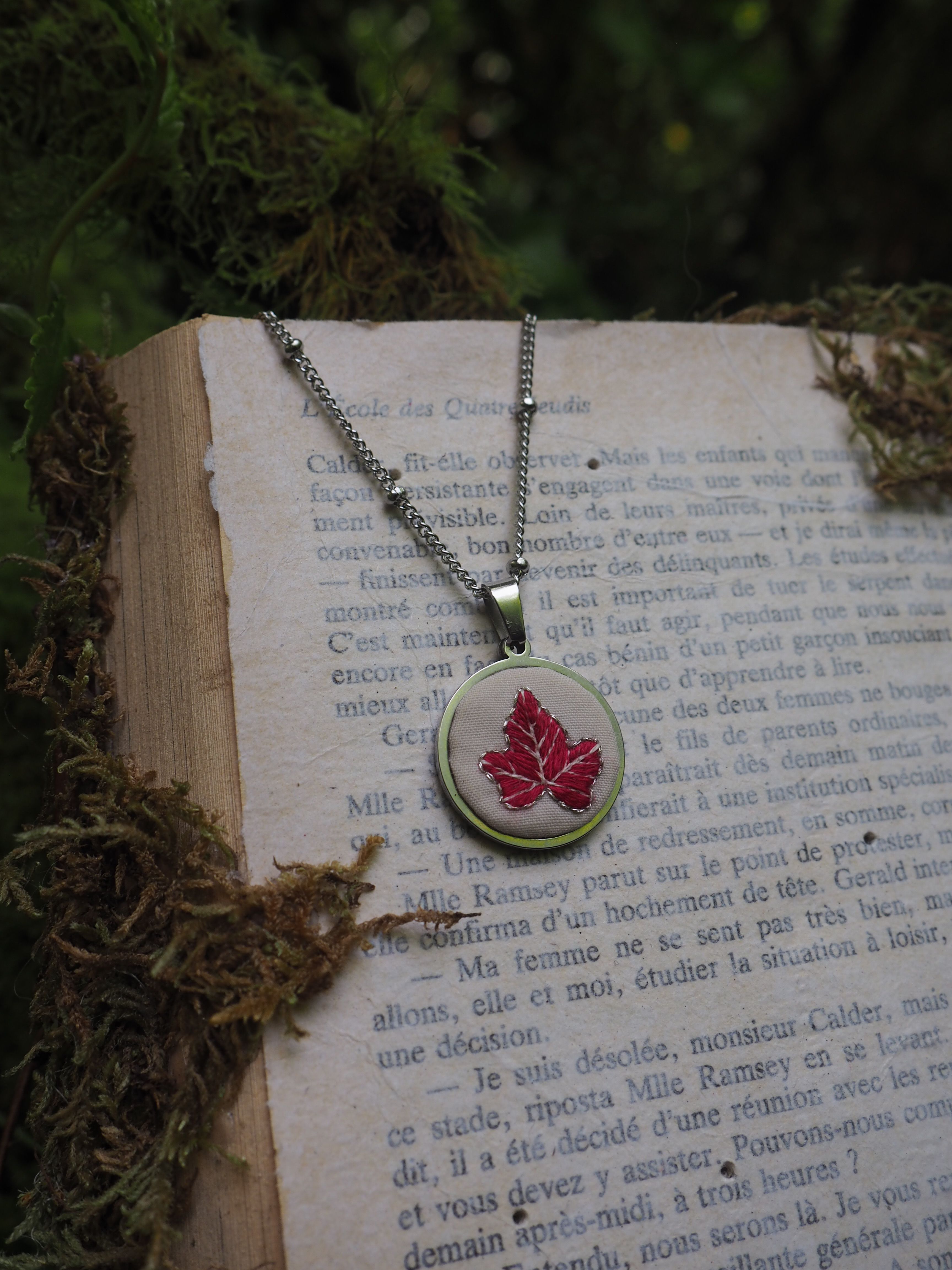 Cette image montre un joli pendentif avec une feuille d'érable rouge sur un fond blanc, suspendu à une chaîne argentée. Le pendentif est posé sur ce qui semble être un livre ou un texte en français. L'arrière-plan montre quelques éléments naturels flous, probablement de la mousse ou des feuilles. L'ambiance de la photo est très poétique et délicate, avec une belle composition qui met en valeur le pendentif et crée une atmosphère littéraire et romantique.