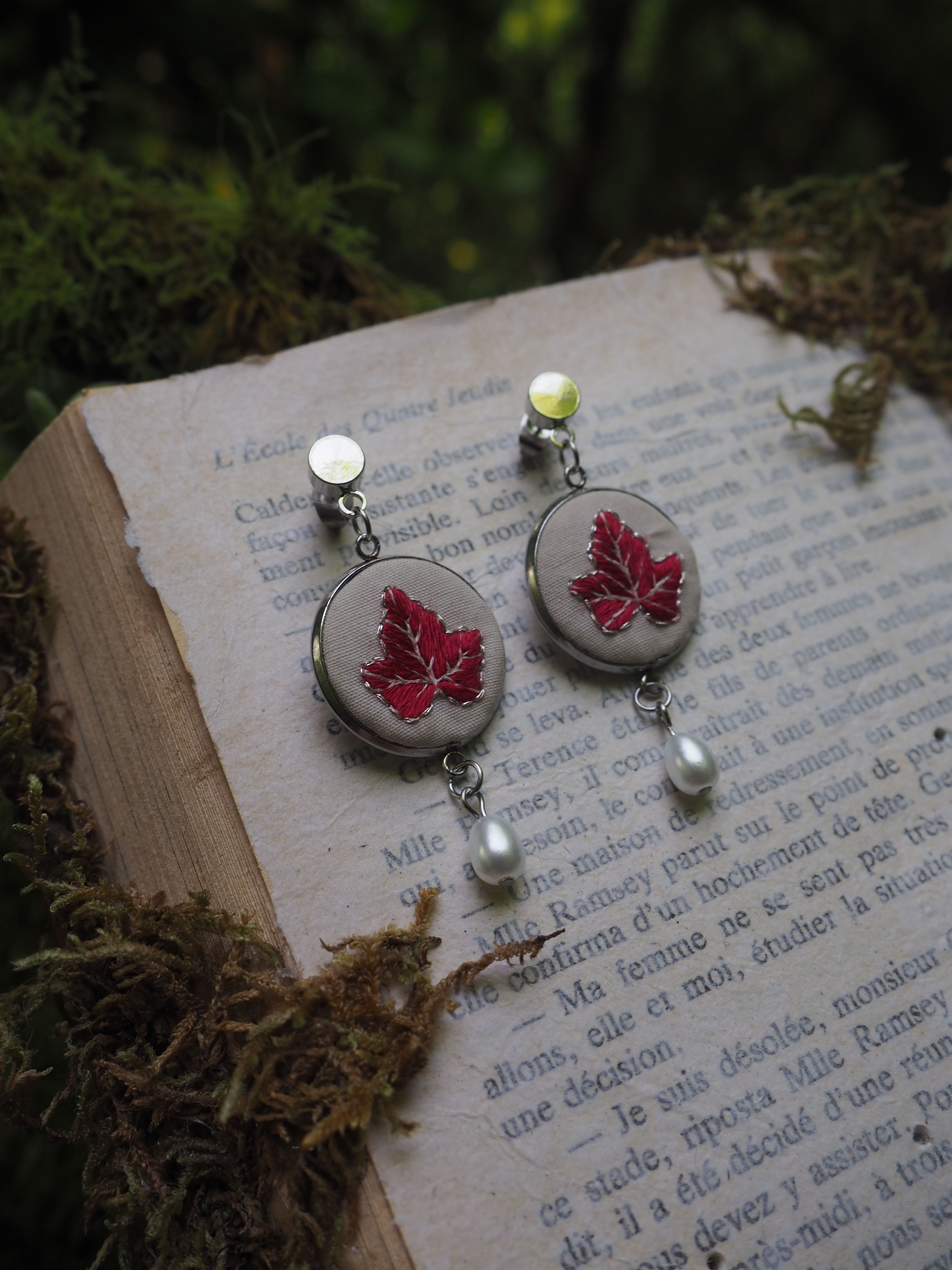 Cette image montre une paire de boucles d'oreilles artisanales posées sur un livre ouvert dans un cadre naturel. Les boucles d'oreilles sont très élégantes, avec des médaillons argentés contenant chacun une feuille d'érable rouge pressée. Au bas de chaque boucle d'oreille pend une petite perle blanche. Les bijoux sont photographiés sur les pages d'un livre en français, avec de la mousse ou des éléments végétaux verts flous en arrière-plan, créant une ambiance très poétique et naturelle. Le design est délicat et évoque l'automne avec les feuilles d'érable rouges.