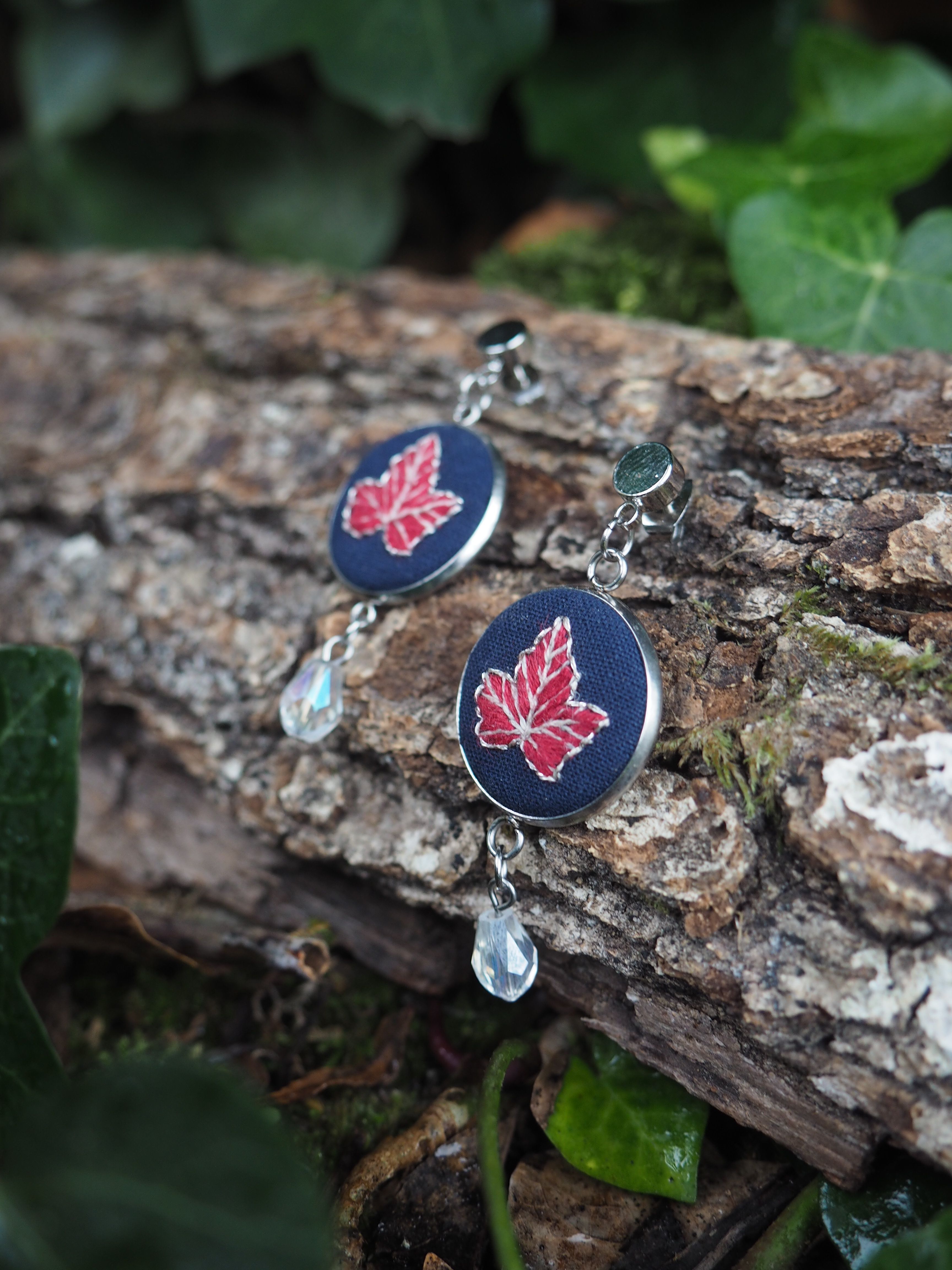Cette image présente une paire de boucles d'oreilles artisanales disposées sur un morceau de bois naturel dans un environnement extérieur verdoyant. **Description des boucles d'oreilles :**
- Base circulaire en tissu ou cuir de couleur bleu marine/noir
- Motif central : une feuille d'érable stylisée de couleur rouge/rose
- Pendentif transparent en forme de goutte suspendu sous chaque cercle
- Crochets métalliques argentés pour l'attache **Environnement :**
- Support : morceau de bois brut avec écorce, texture naturelle visible
- Arrière-plan : végétation verte floue créant un effet de profondeur
- Feuilles vertes visibles en arrière-plan
- Éclairage naturel doux qui met en valeur les détails **Style artistique :**
- Photographie en gros plan avec une faible profondeur de champ
- Composition équilibrée mettant en valeur l'artisanat
- Contraste harmonieux entre les couleurs sombres des boucles et le vert naturel
- Ambiance naturelle et organique qui complète le style artisanal des bijoux L'ensemble évoque un travail d'artisanat soigné avec une esthétique inspirée de la nature, particulièrement du symbole canadien de la feuille d'érable.
