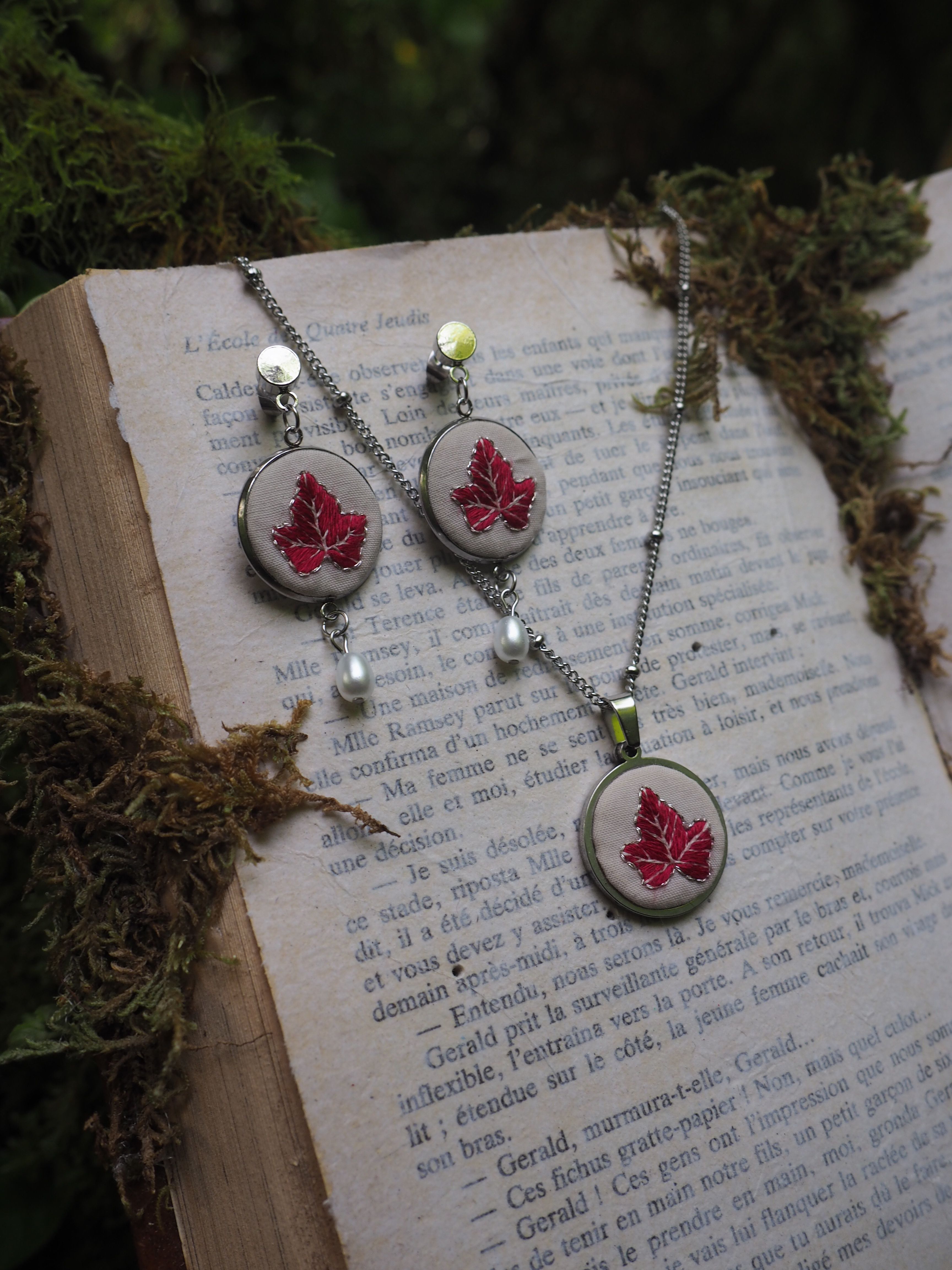Cette image montre un ensemble de bijoux artisanaux délicats composé de boucles d'oreilles et d'un collier assortis. Chaque pièce présente une feuille d'érable rouge brodée sur un fond blanc, encadrée dans ce qui semble être des supports métalliques circulaires. Les bijoux sont photographiés sur les pages ouvertes d'un livre ancien, avec de la mousse ou des éléments naturels verts disposés autour. Les boucles d'oreilles et le pendentif sont ornés de petites perles blanches et de ce qui semble être des perles de verre vert clair. L'ensemble dégage une atmosphère romantique et naturelle, avec un style vintage délicat.