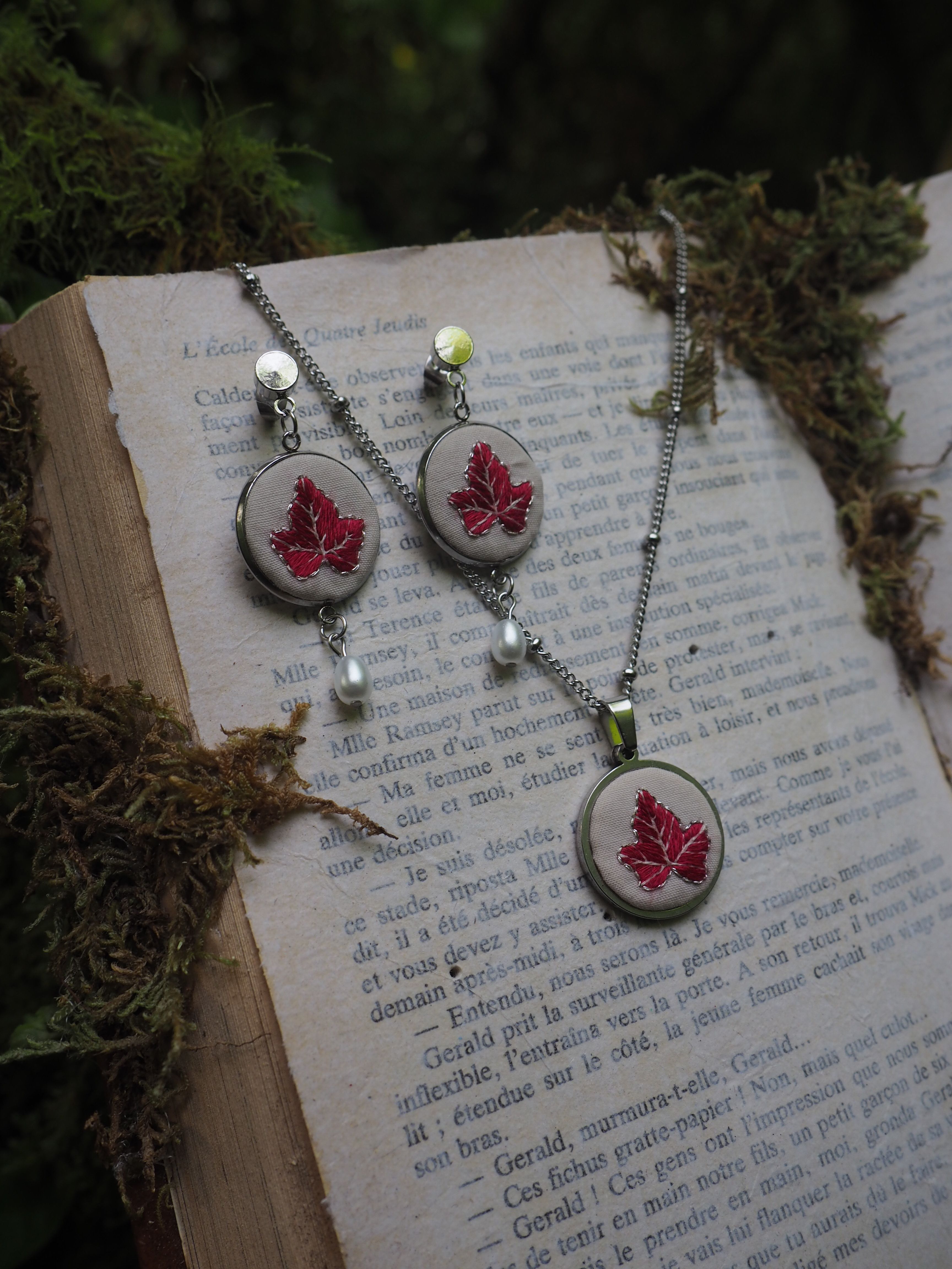 Cette image montre un ensemble de bijoux élégants composé de boucles d'oreilles et d'un collier assortis, disposés sur les pages d'un livre ancien. Les bijoux présentent un motif de feuille d'érable rouge dans des médaillons argentés. Le design est agrémenté de petites perles blanches et de ce qui semble être des perles de verre vert clair. L'arrière-plan montre du texte imprimé sur les pages du livre et quelques éléments de mousse ou de végétation, créant une ambiance naturelle et romantique. La composition est artistique et évoque un style vintage ou fait main.