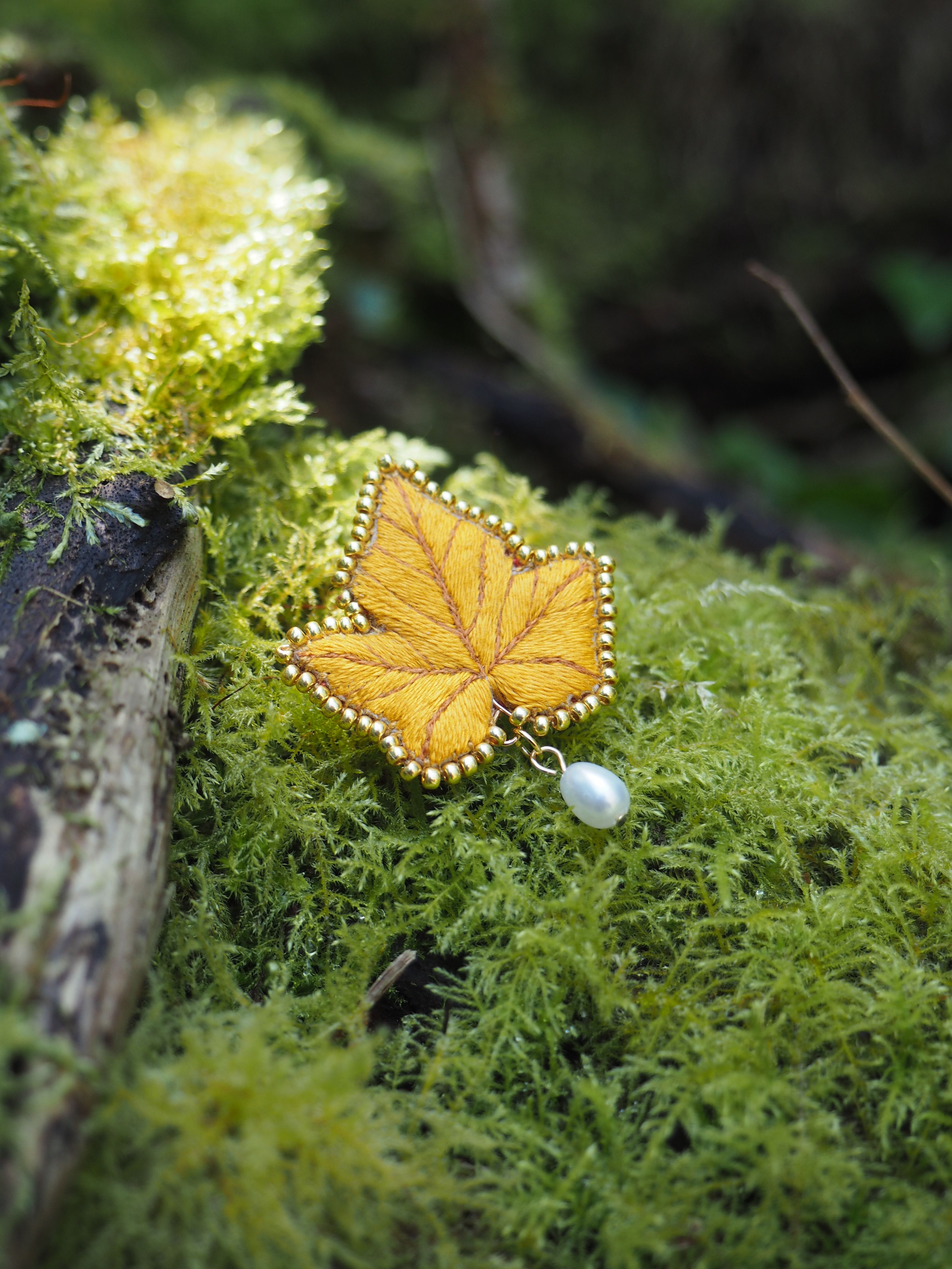 Cette image montre une délicate broche ou un ornement en forme de feuille d'érable, réalisé en ce qui semble être du métal doré ou du tissu brodé. La broche est posée sur un lit de mousse verte luxuriante, créant un beau contraste entre le doré de la feuille et le vert vif de la mousse. Les bords de la broche sont ornés de petites perles blanches qui forment une bordure décorative. L'arrière-plan est flou mais naturel, suggérant un cadre forestier. La composition est très artistique et évoque une ambiance féerique ou enchantée, comme si on avait découvert un trésor délicat dans la nature.