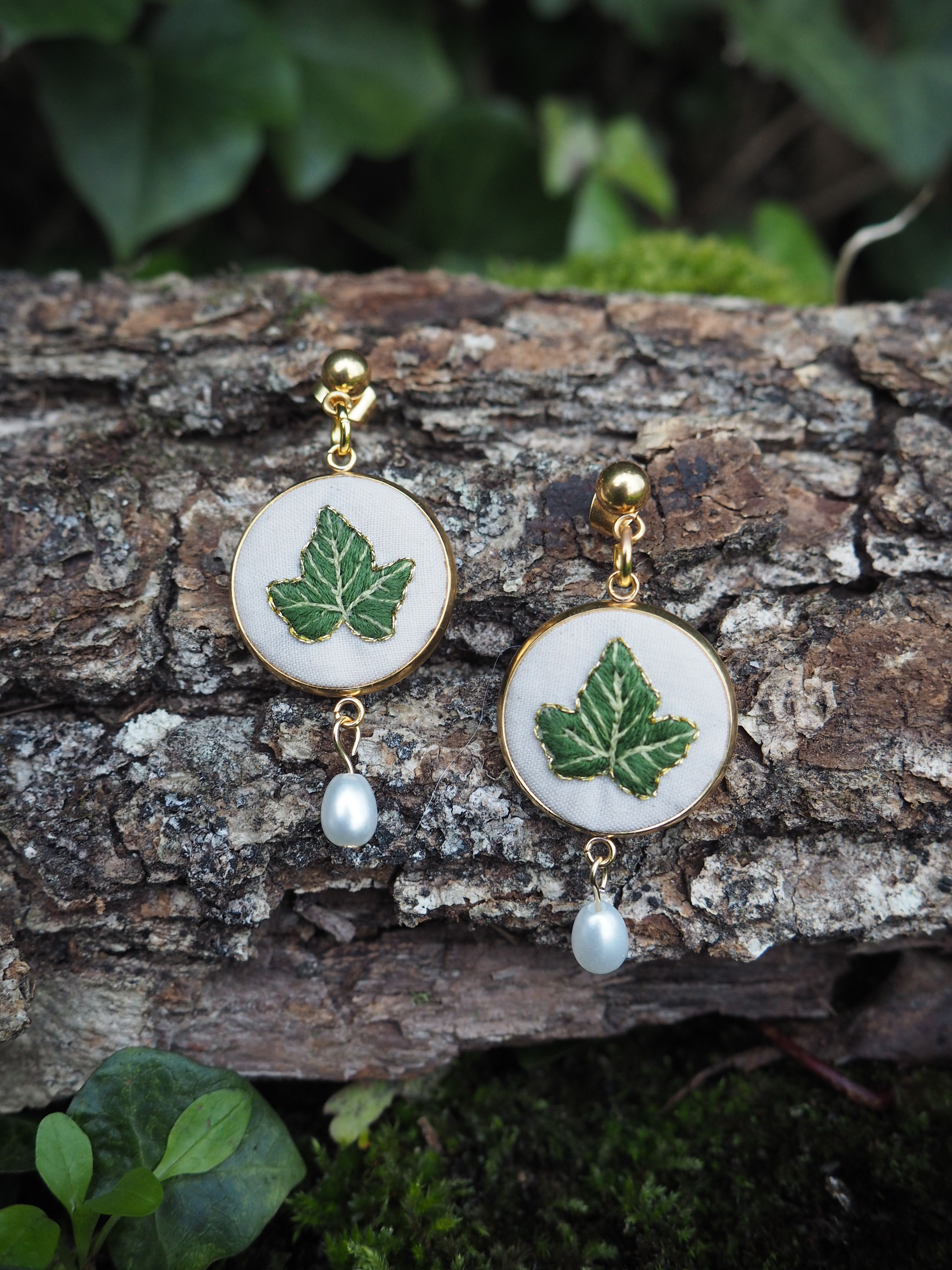 Cette image montre une paire de boucles d'oreilles artisanales élégantes posées sur un morceau d'écorce d'arbre. Les boucles d'oreilles sont composées de médaillons blancs circulaires sur lesquels sont peintes des feuilles d'érable vertes. Chaque boucle d'oreille est ornée d'une petite perle blanche qui pend en dessous du médaillon. Les attaches semblent être en métal doré. L'arrière-plan naturel avec l'écorce texturée et quelques feuilles vertes floues met parfaitement en valeur ces bijoux délicats qui allient nature et élégance.