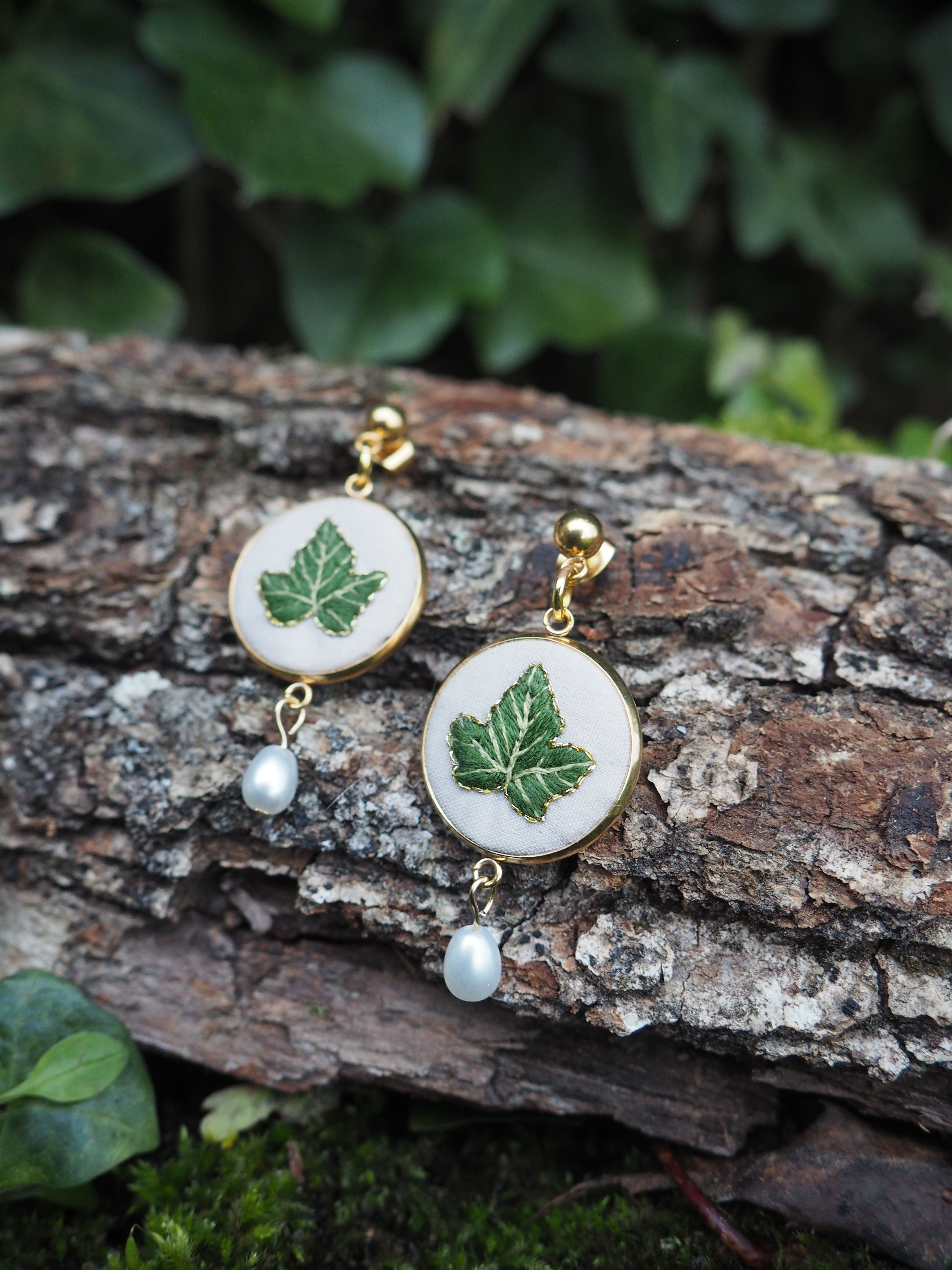 Cette image montre une paire de boucles d'oreilles artisanales délicates posées sur un morceau d'écorce d'arbre. Les boucles d'oreilles sont composées de médaillons blancs circulaires sur lesquels sont peintes des feuilles d'érable vertes. Chaque boucle d'oreille est ornée d'une petite perle blanche qui pend en dessous du médaillon. Les attaches semblent être en métal doré. L'arrière-plan montre du feuillage vert flou, créant une ambiance naturelle et forestière qui complimente parfaitement le motif de feuille d'érable sur les bijoux.