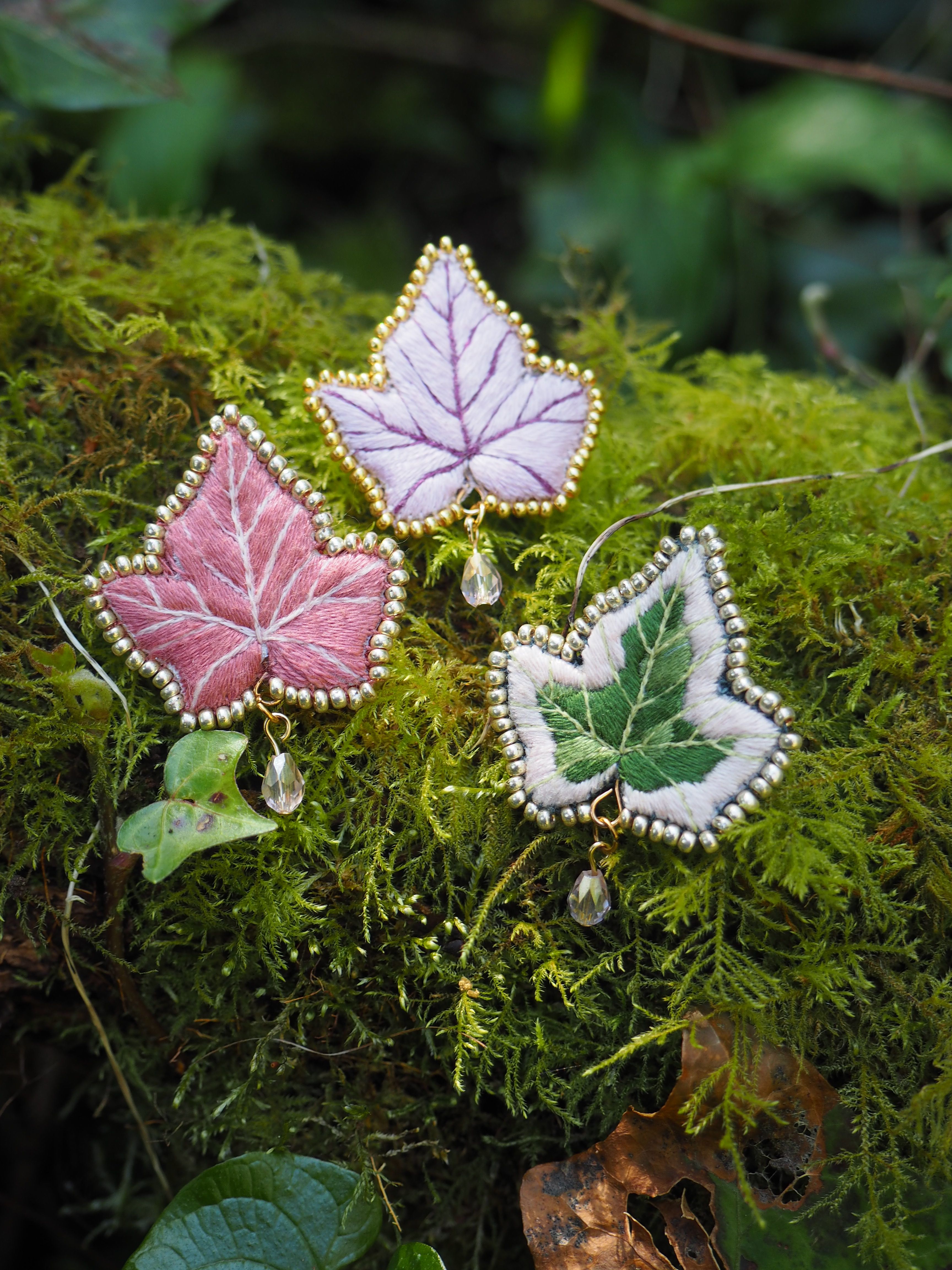 Cette image montre trois jolies broches ou pendentifs en forme de feuilles d'érable, posées sur un lit de mousse verte. Les feuilles sont délicatement confectionnées avec des bordures argentées et sont de trois couleurs différentes : rose, lavande et vert pâle. Chaque feuille semble être ornée d'une petite perle ou cristal suspendu. Le contraste entre les bijoux délicats et la mousse naturelle crée une ambiance féerique et enchantée. L'arrière-plan est flou mais on peut apercevoir d'autres éléments naturels comme des feuilles vertes. C'est une composition très artistique qui mélange élégamment des éléments de joaillerie avec la nature.
