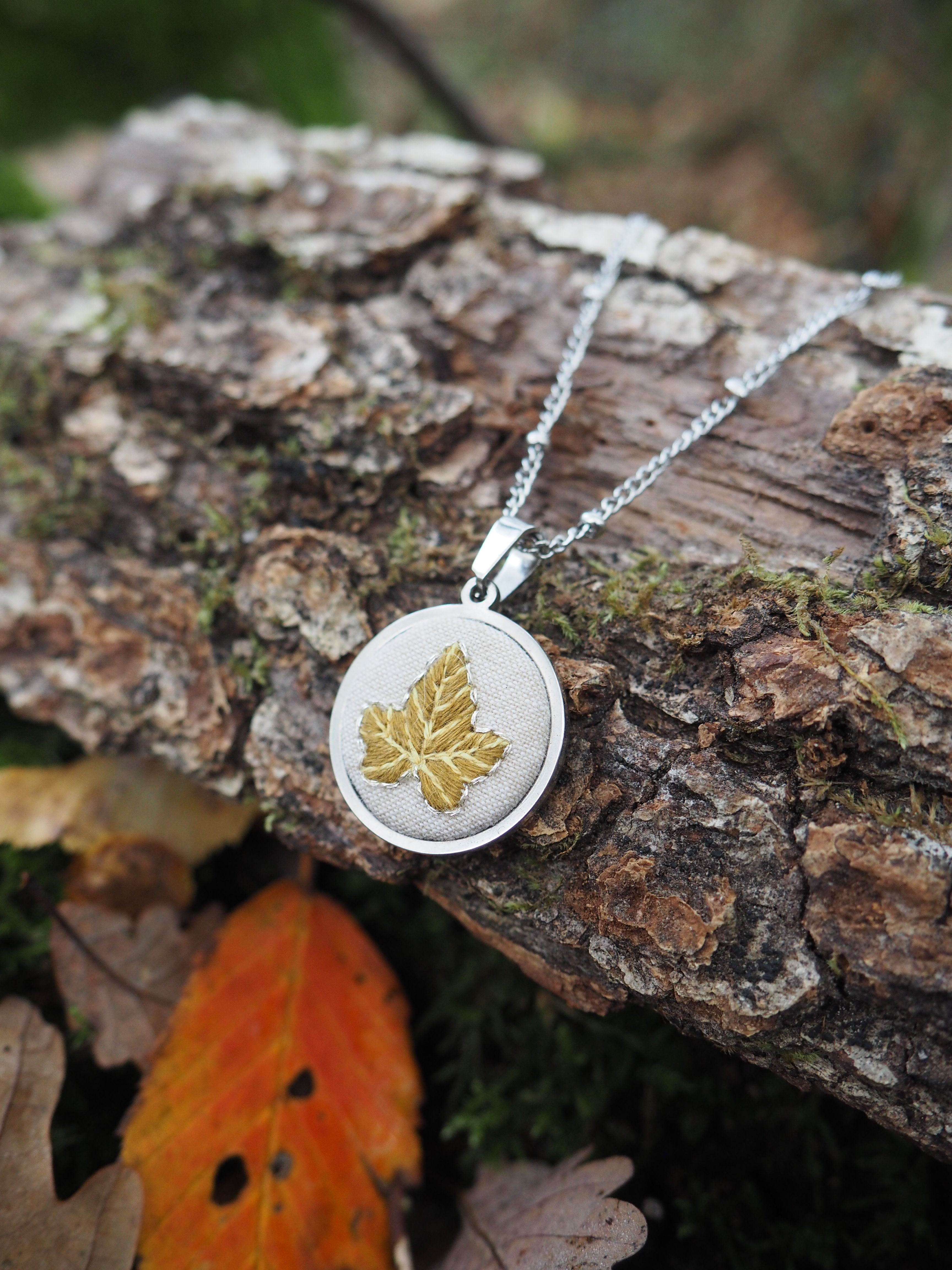 Cette image présente un bijou élégant disposé dans un cadre naturel automnal. Au centre, on peut voir un pendentif circulaire en métal argenté suspendu à une chaîne fine. Le pendentif arbore un motif délicat de feuille dorée gravée ou incrustée, créant un joli contraste entre l'argent et l'or. Le collier repose sur un morceau de bois flotté ou une branche d'arbre aux tons bruns et gris, dont l'écorce rugueuse et texturée offre un arrière-plan naturel parfait. L'environnement suggère une scène forestière ou de sous-bois, avec de la mousse verdâtre visible sur le bois et quelques feuilles d'automne aux couleurs chaudes - notamment une feuille orange vif au premier plan. La composition photographique met en valeur le contraste entre l'objet manufacturé raffiné et l'environnement naturel brut, créant une esthétique harmonieuse qui évoque les thèmes de la nature et de l'artisanat. L'éclairage doux fait ressortir les détails du pendentif tout en préservant l'ambiance naturelle de la scène.