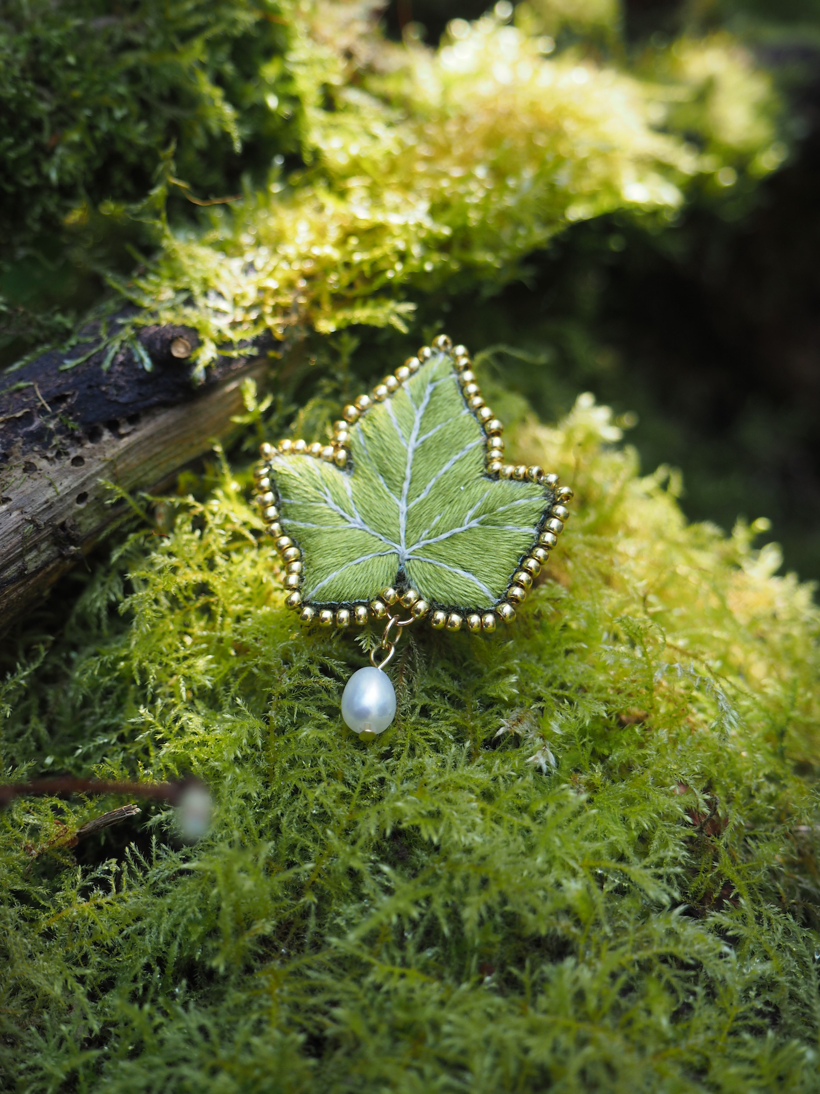 Cette image montre un magnifique bijou en forme de feuille posé sur un lit de mousse verte. Le bijou semble être une broche ou un pendentif qui représente une feuille d'érable délicatement ouvragée en vert, bordée de petites perles dorées. Une perle blanche nacrée est suspendue en dessous de la feuille. L'arrière-plan est composé d'une mousse luxuriante d'un vert vif, créant un cadre naturel parfait pour mettre en valeur ce bijou délicat. La photographie capture bien les détails de l'artisanat et crée une ambiance féerique et naturelle avec la combinaison du bijou et de son environnement moussu.