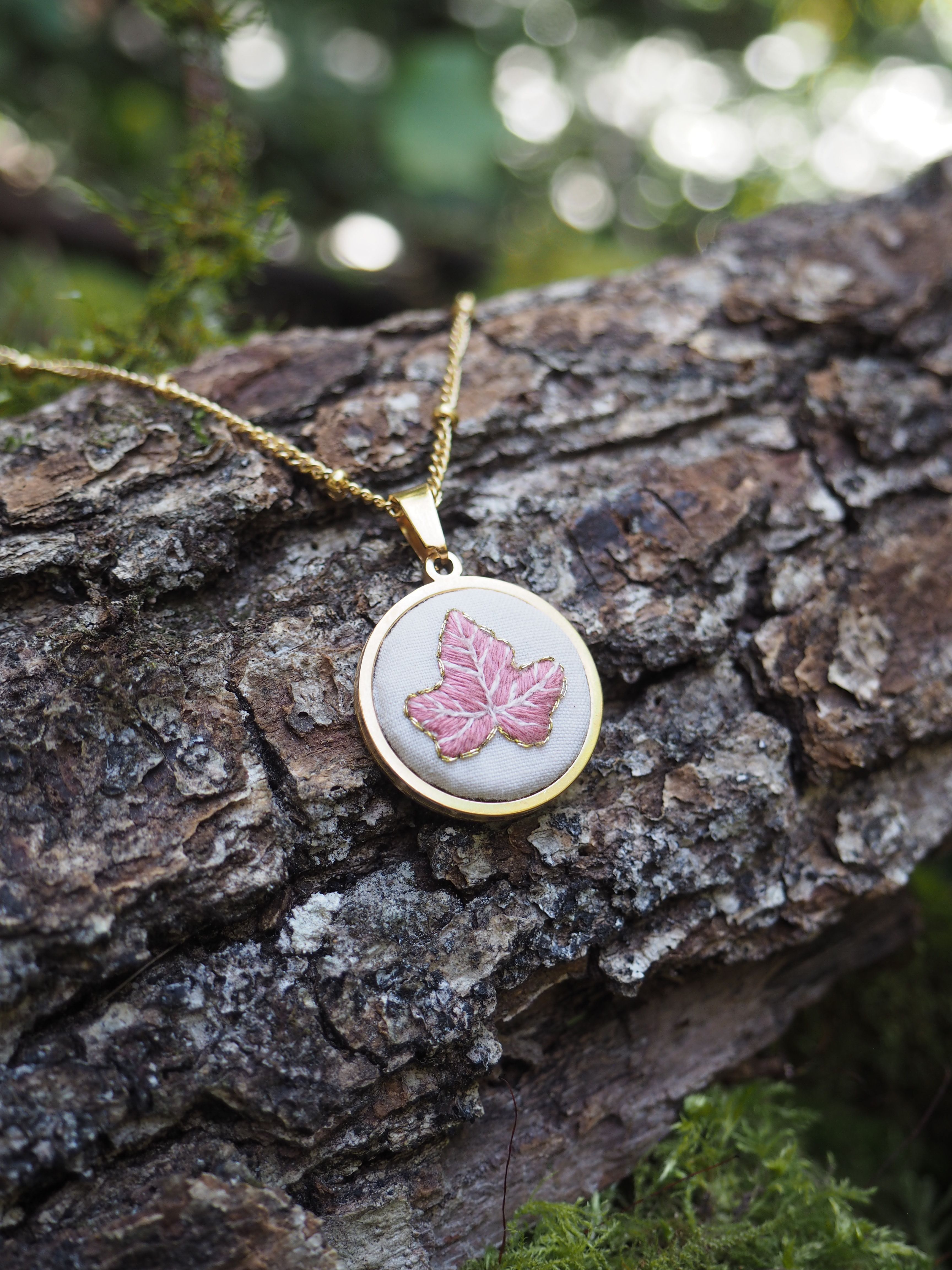 Cette image montre un délicat pendentif posé sur un tronc d'arbre couvert d'écorce rugueuse. Le pendentif est rond, monté sur une chaîne dorée, et présente un motif de feuille d'érable rose sur un fond clair. La composition est artistique avec un bel effet de bokeh (flou) en arrière-plan créant des points de lumière verts et doux. Il y a également un peu de mousse verte visible sur l'écorce, ajoutant à l'ambiance naturelle et forestière de la photo. Le contraste entre le bijou délicat et la texture rugueuse de l'écorce crée une belle composition photographique.