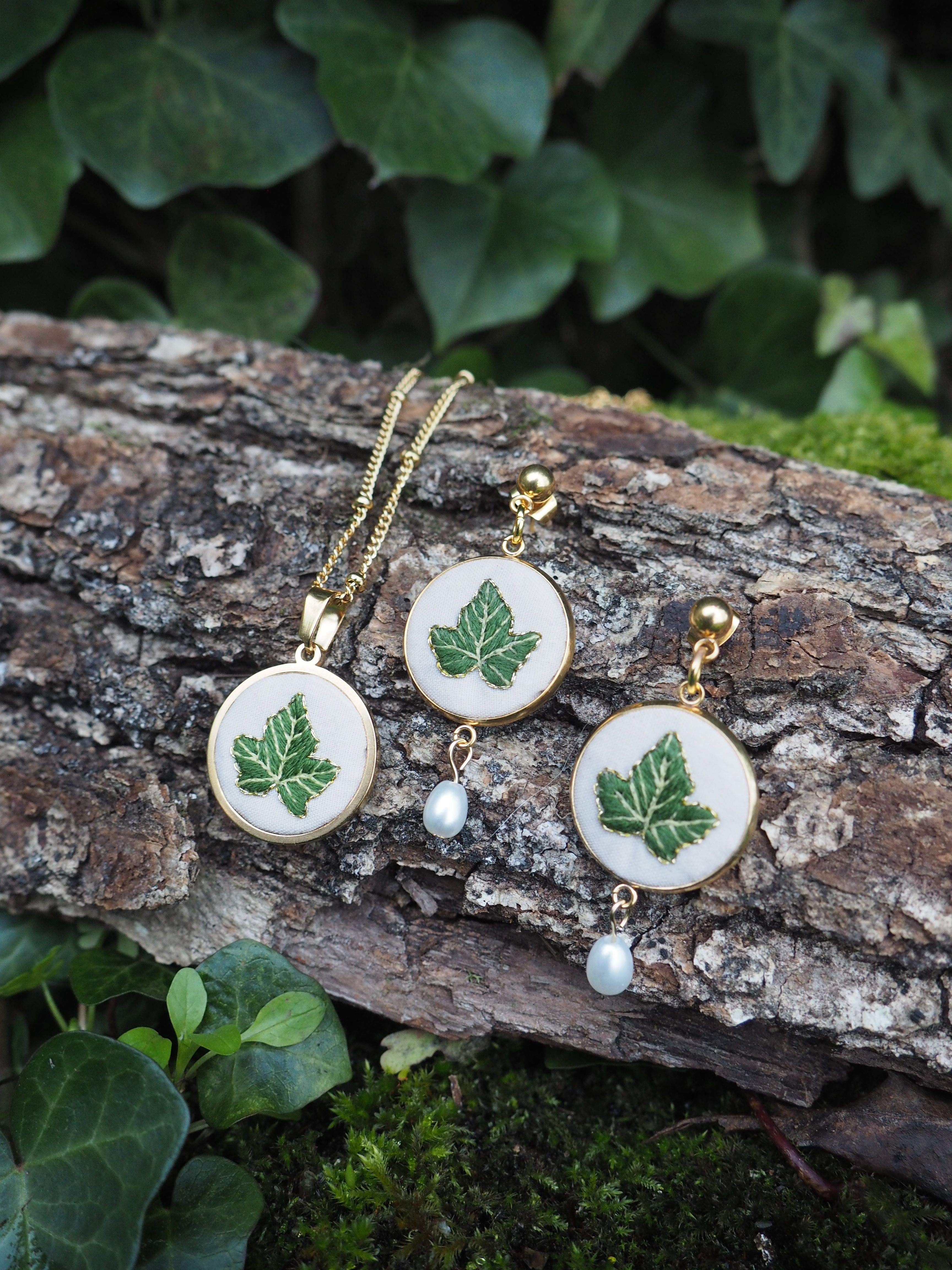 Cette image montre un ensemble de bijoux délicats posés sur un morceau d'écorce d'arbre avec de la mousse et du lierre en arrière-plan. Les bijoux semblent être un collier et des boucles d'oreilles assortis, fabriqués dans un style nature. Chaque pièce présente un médaillon rond avec une feuille verte peinte ou incrustée sur un fond blanc, et est ornée d'une petite perle suspendue. Les montures sont de couleur dorée. La composition est très naturelle et harmonieuse, mettant en valeur le thème botanique des bijoux en les photographiant dans un environnement naturel avec des feuilles vertes et de l'écorce texturée.