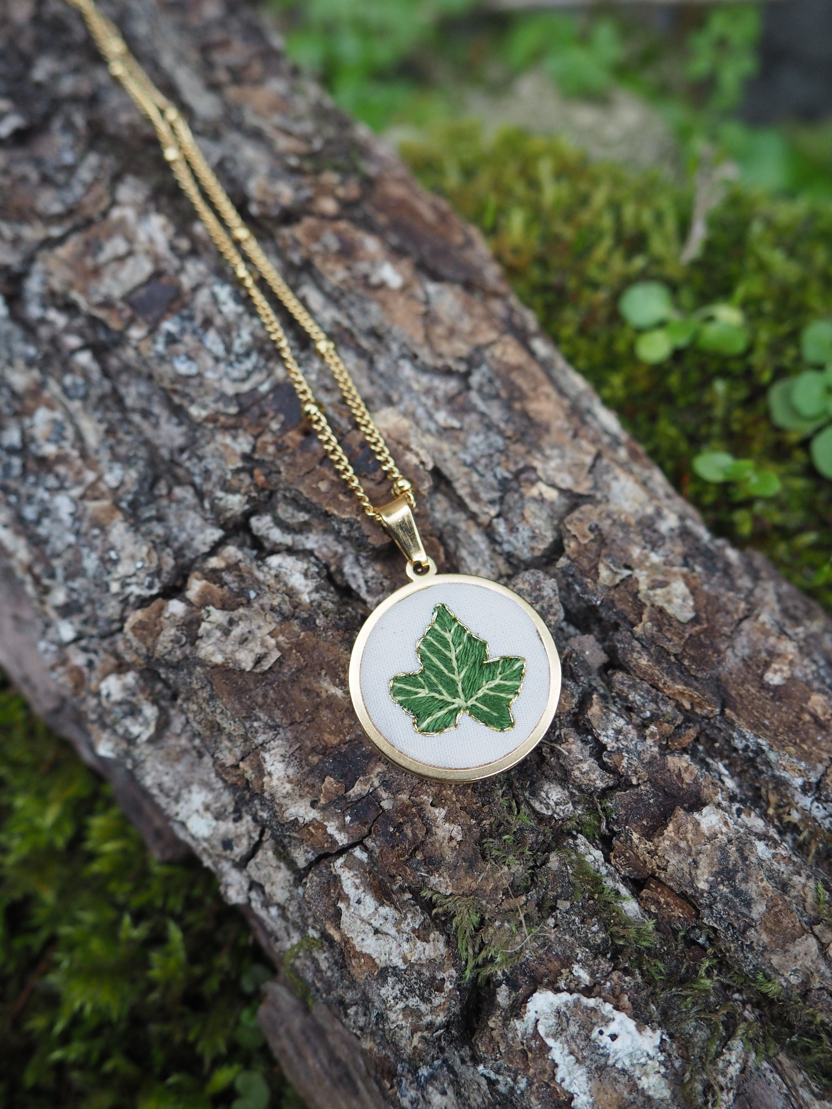 Cette image montre un délicat pendentif sur une chaîne dorée, posé sur un morceau de bois couvert de mousse. Le pendentif est de forme ronde et semble être en émail blanc ou en porcelaine, décoré d'une feuille d'érable verte. La composition est très naturelle et harmonieuse, avec le contraste entre le bijou raffiné et la texture rugueuse du bois et la mousse verte tendre. C'est une belle mise en scène qui évoque la nature et l'artisanat.