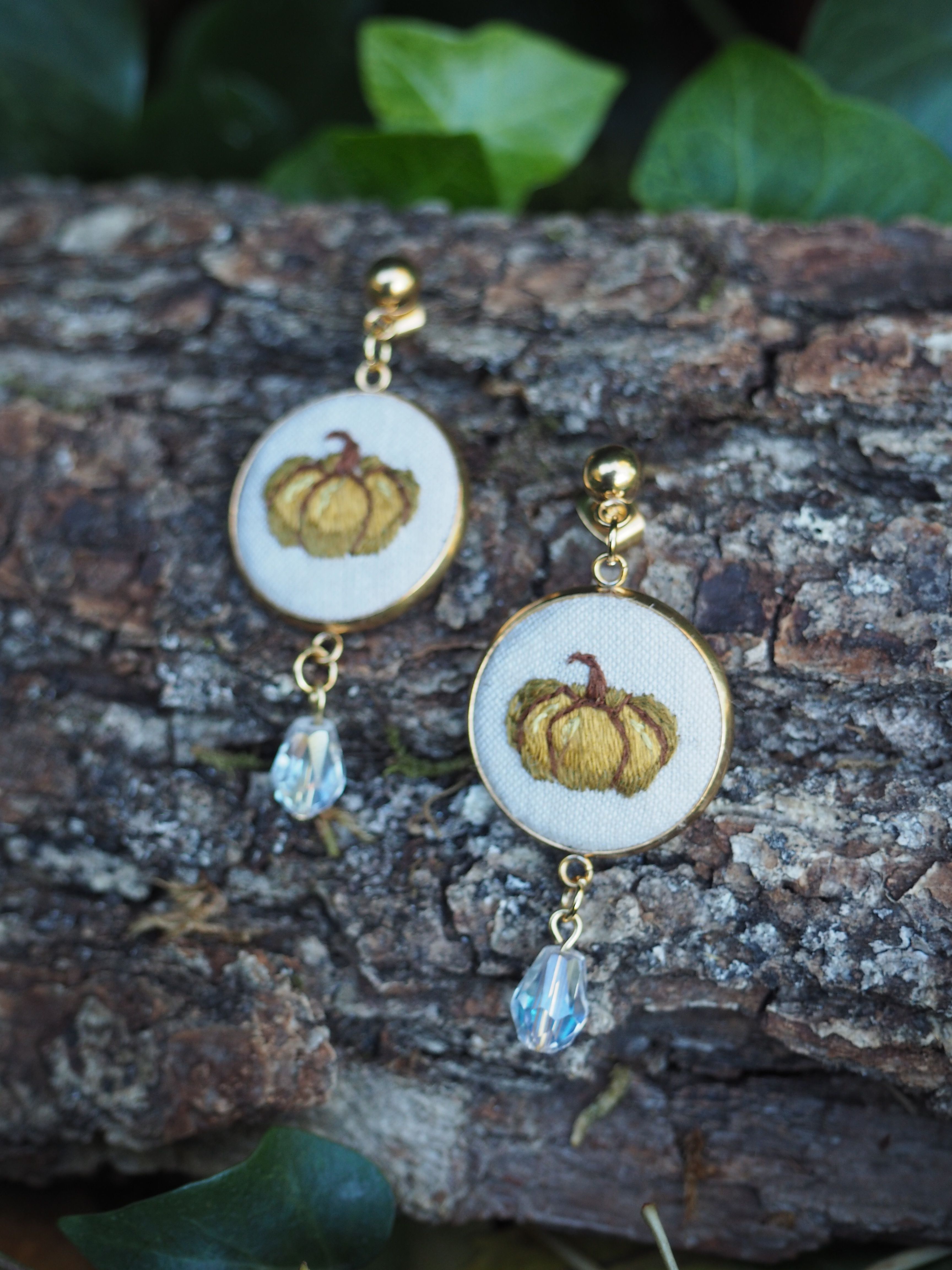 Cette image présente une paire de boucles d'oreilles élégantes disposées sur une surface naturelle en bois brut.  **Caractéristiques des bijoux :**
- **Design principal** : Chaque boucle d'oreille comporte un médaillon ovale blanc avec un motif décoratif doré représentant ce qui semble être une citrouille ou un fruit stylisé
- **Finitions** : Les éléments métalliques sont dorés, donnant un aspect luxueux
- **Pendentifs** : Chaque boucle se termine par une petite perle ou cristal transparent qui accroche la lumière
- **Style** : L'ensemble évoque un style vintage ou artisanal avec une touche automnale **Mise en scène :**
- Les boucles d'oreilles reposent sur une écorce d'arbre texturée, créant un contraste naturel
- L'arrière-plan flou laisse apercevoir quelques feuilles vertes
- L'éclairage naturel met en valeur les détails dorés et la transparence des pendentifs Cette composition photographique met parfaitement en valeur ces bijoux artisanaux dans un cadre naturel et organique.