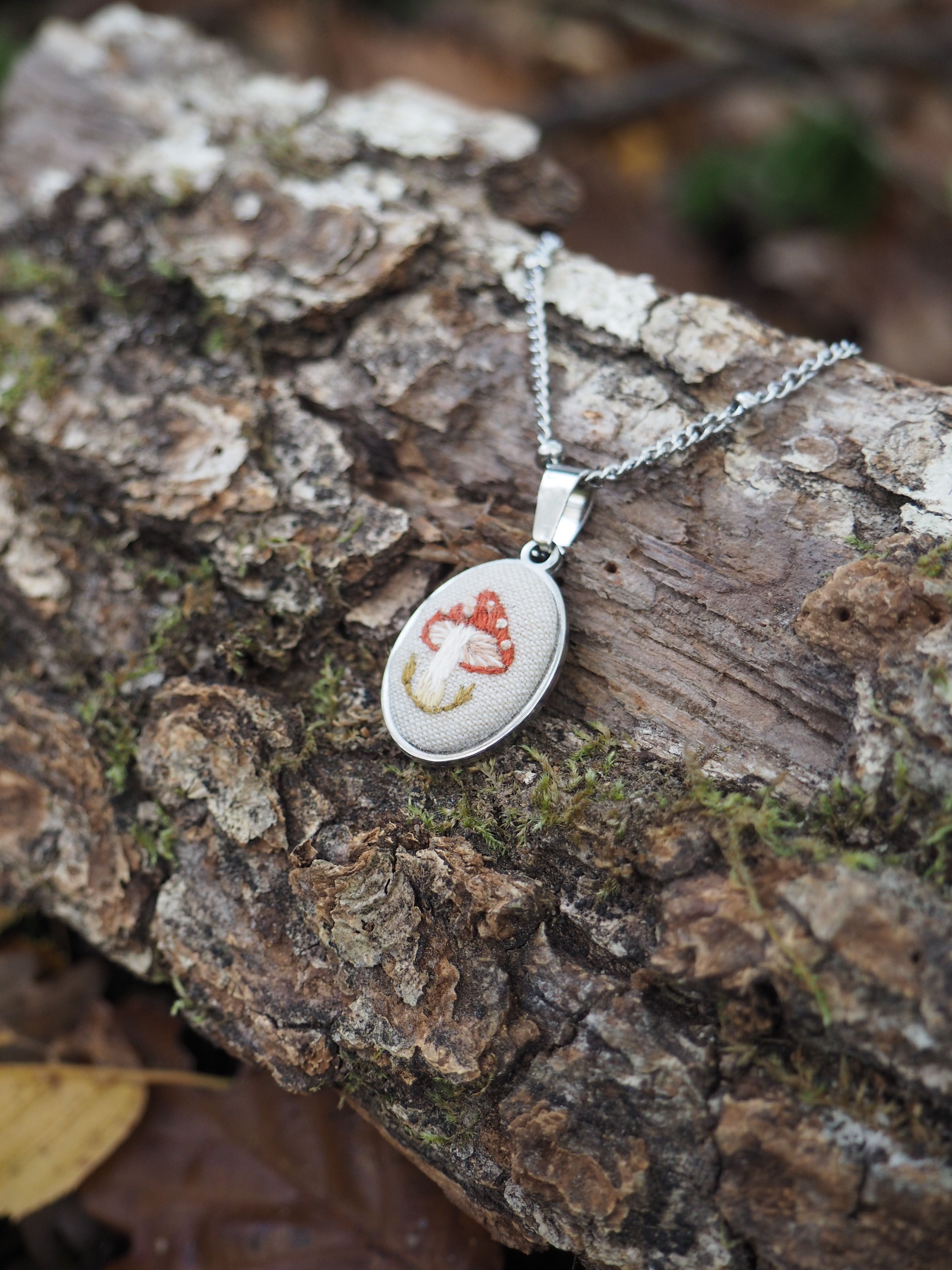 Cette image présente un collier élégant posé sur un morceau de bois naturel. Le bijou se compose d'une chaîne argentée fine et délicate qui supporte un pendentif ovale. Le pendentif lui-même semble être serti d'argent et contient ce qui apparaît être une pierre semi-précieuse ou un minéral avec des motifs naturels dans des tons de blanc et de rouge-orange, créant des veines et des marbrures organiques caractéristiques. Le bois sur lequel repose le collier présente une texture rugueuse et patinée, avec des traces de mousse verdâtre qui témoignent de son exposition aux éléments naturels. L'écorce montre différentes nuances de brun et de gris, avec des fissures et des aspérités qui créent un contraste intéressant avec la finesse du bijou. L'ensemble de la composition évoque un style naturel et artisanal, où la beauté brute du bois fait ressortir l'élégance travaillée du pendentif. L'éclairage doux met en valeur les détails de la pierre et le brillant de la monture argentée.