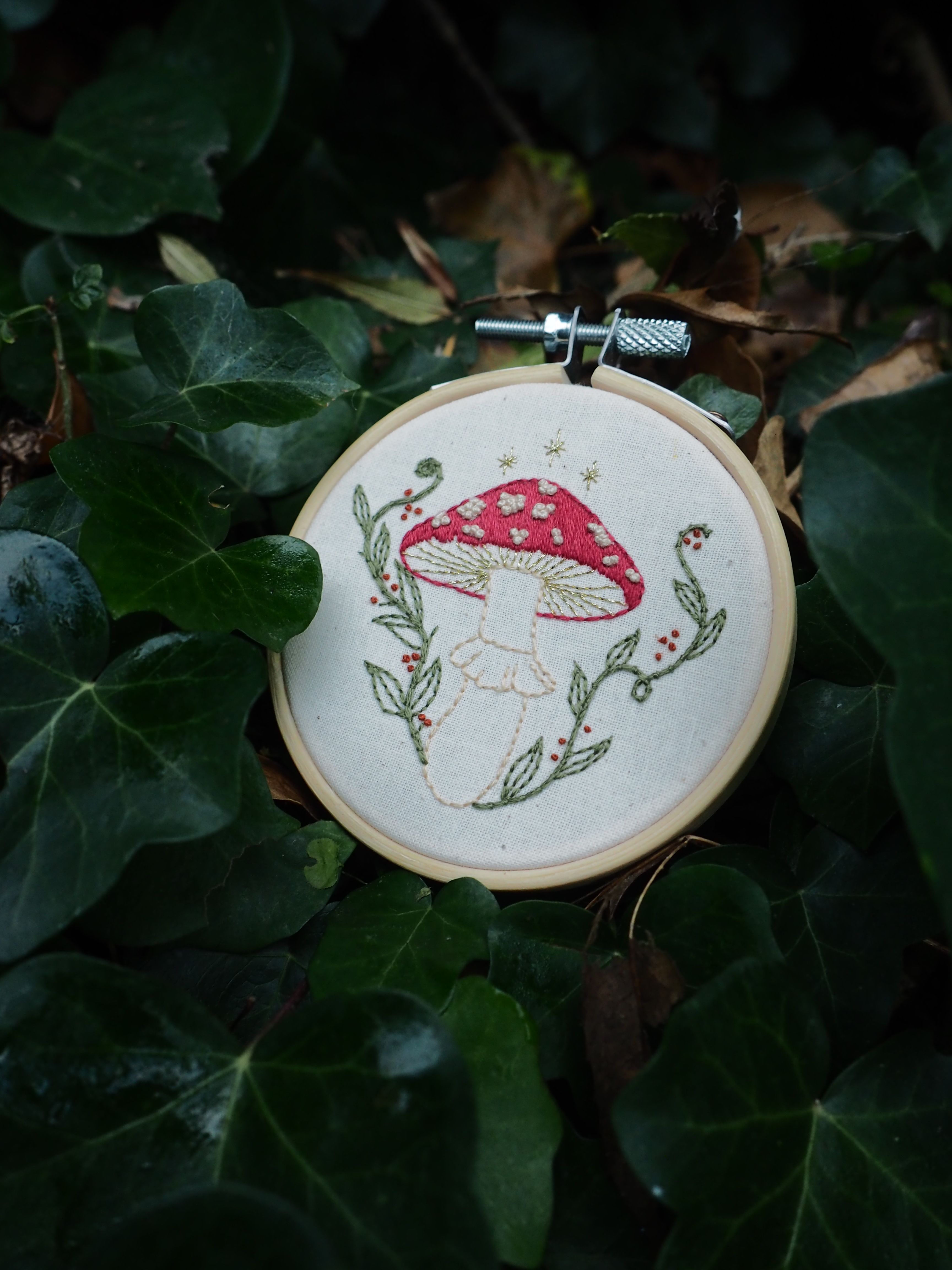 Cette image présente une charmante broderie artisanale disposée dans un environnement naturel. Voici ce que je peux observer : **L'objet principal :**
- Un tambour à broder circulaire en bois contenant une broderie sur tissu blanc
- Le motif brodé représente un champignon rouge à pois blancs (probablement une amanite tue-mouches) avec sa tige blanche
- Le champignon est entouré de délicates branches et feuillages brodés en vert
- Le travail de broderie semble fait à la main avec différents points et couleurs **Le cadre naturel :**
- Le tambour est posé sur un lit de feuilles vertes, créant un contraste harmonieux
- L'environnement semble être de la végétation dense, possiblement du lierre ou des feuilles similaires
- L'éclairage naturel met en valeur les détails de la broderie
- La composition crée un lien poétique entre l'art textile et la nature Cette mise en scène évoque l'univers des contes de fées et souligne le lien entre l'artisanat traditionnel et les motifs inspirés de la nature. La qualité artistique de la broderie et sa présentation dans ce cadre végétal créent une atmosphère à la fois nostalgique et enchantée.