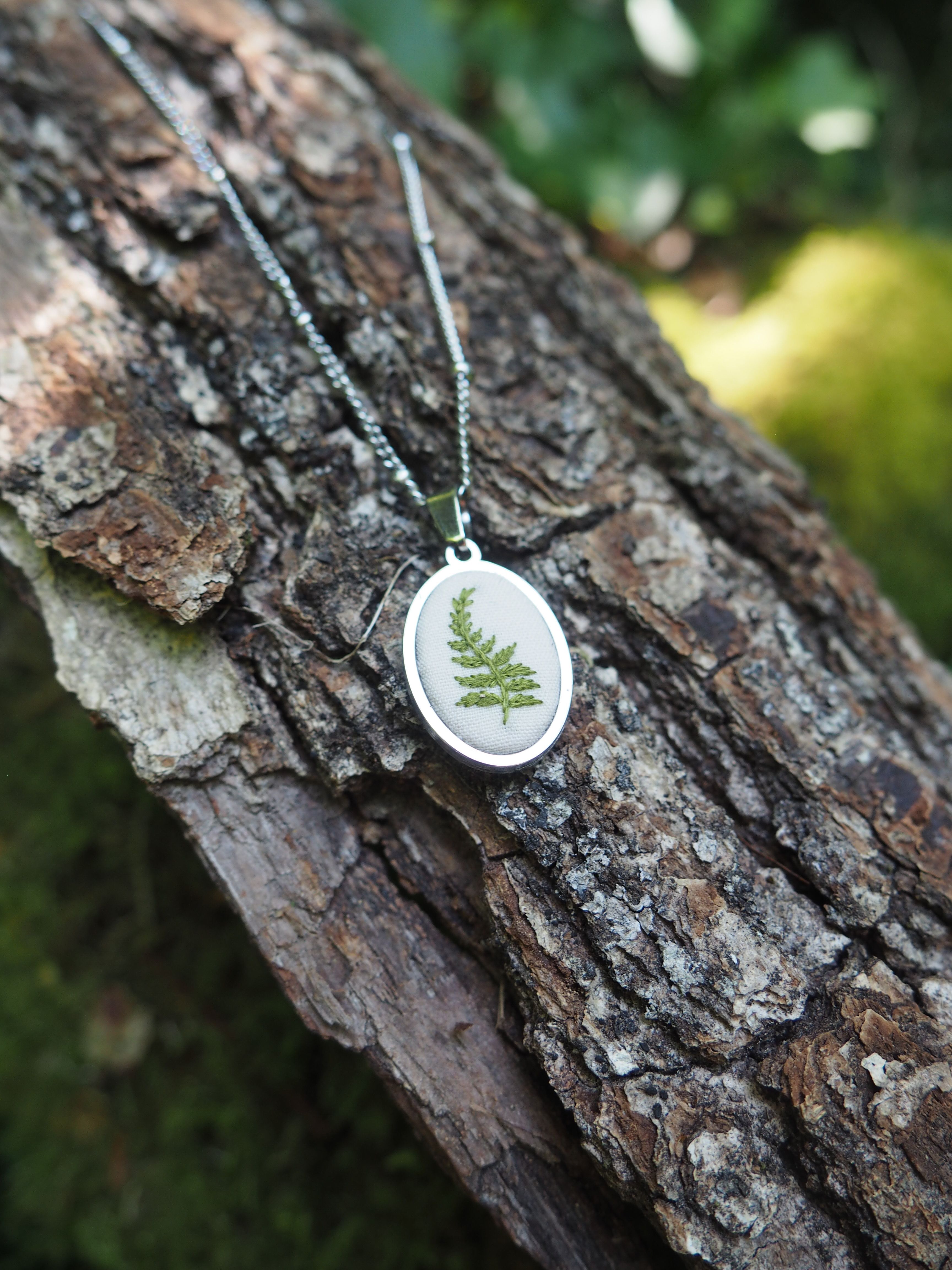 L'image montre des bijoux ou des pendentifs délicats qui semblent contenir de véritables petites fougères préservées. Ces pendentifs sont de forme ronde avec ce qui semble être un cadre blanc ou argenté, et à l'intérieur se trouve une fougère verte naturelle, probablement conservée sous verre ou dans de la résine. Ils sont photographiés sur ce qui semble être l'écorce rugueuse d'un arbre, créant un contraste intéressant entre l'aspect naturel brut de l'écorce et la délicatesse des pendentifs. L'image a une belle composition naturelle et organique qui met en valeur ces bijoux botaniques.