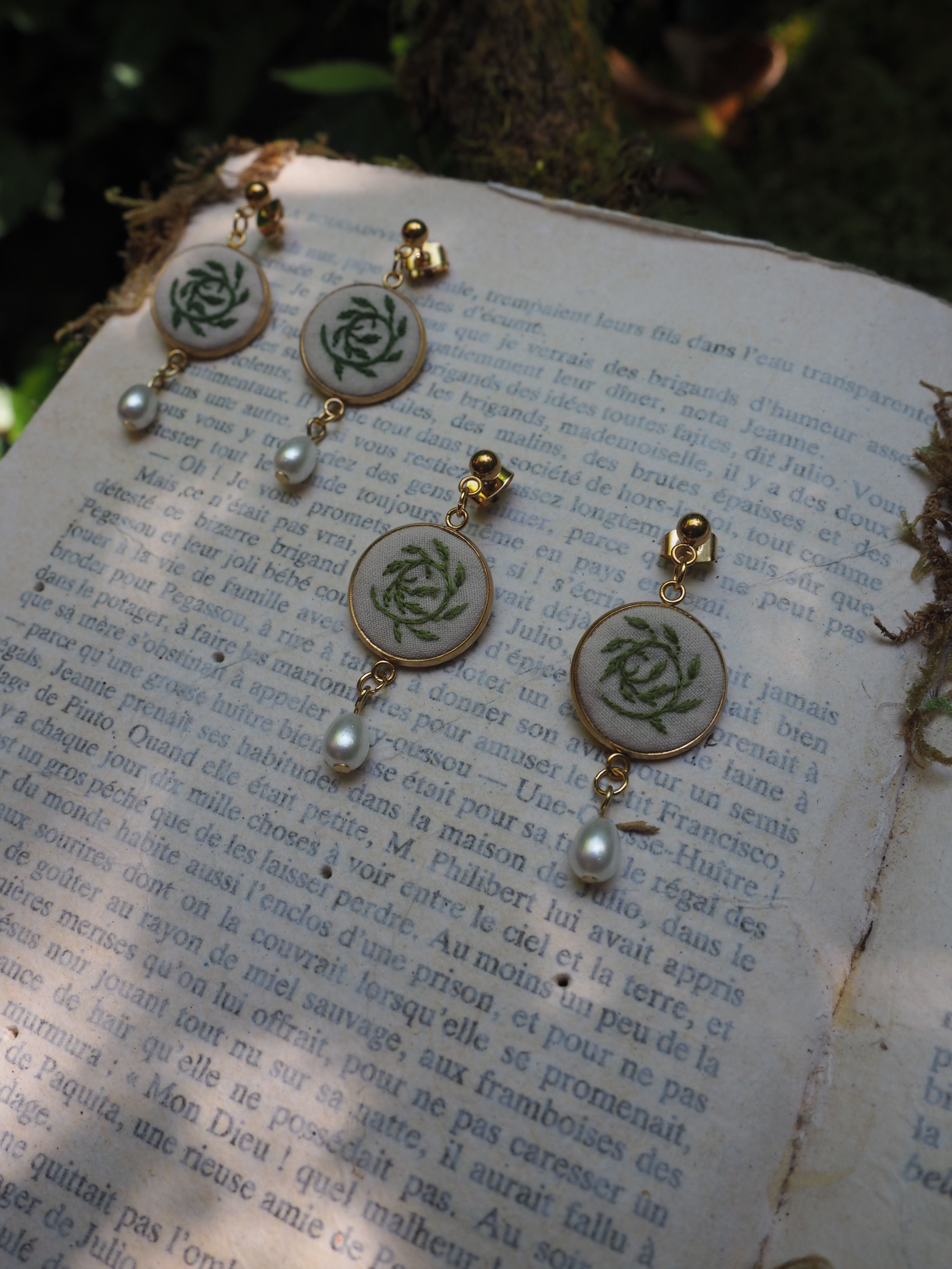 Cette image montre des bijoux délicats posés sur ce qui semble être une page de livre en français. Les bijoux sont des boucles d'oreilles qui présentent un motif de rose ou un motif floral circulaire en vert sur un fond clair, probablement de la porcelaine ou de l'émail. Chaque boucle d'oreille est ornée d'une petite perle blanche qui pend au bas. Les bijoux ont une apparence vintage ou artisanale, avec des détails dorés. Ils sont photographiés sur une page de texte qui semble être d'un livre ancien ou vintage, créant une composition romantique et nostalgique.