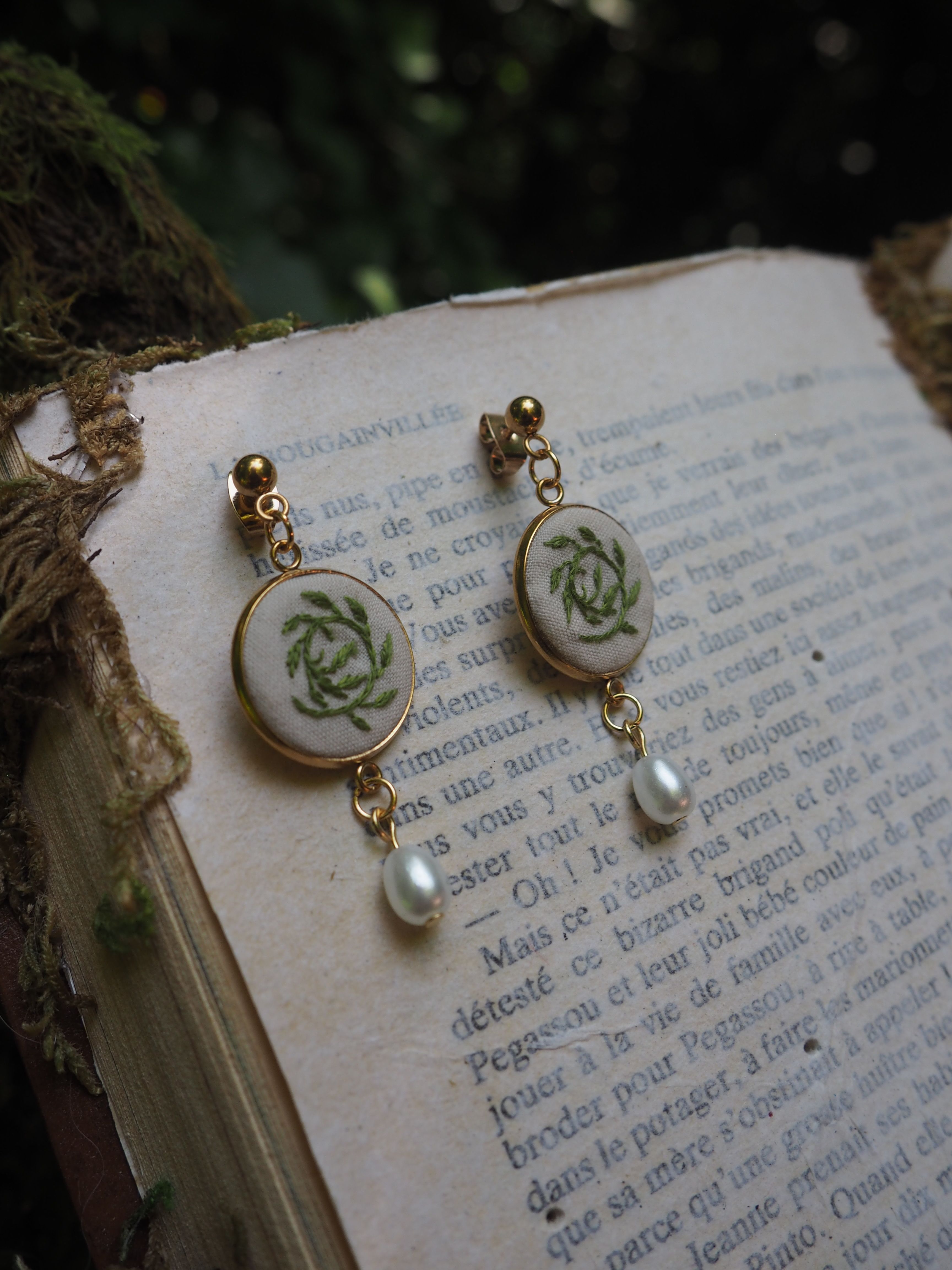 Cette image montre des boucles d'oreilles délicates posées sur ce qui semble être une page de livre en français. Les boucles d'oreilles paraissent être faites avec des petits médaillons en porcelaine ou en céramique, décorés de motifs floraux verts pâles, possiblement des roses. Elles sont agrémentées de perles et montées sur ce qui semble être des attaches dorées. Le fond montre du texte imprimé en français sur une page qui a un aspect vieilli ou vintage. L'image a une qualité romantique et vintage qui rappelle un style shabby chic ou français ancien.