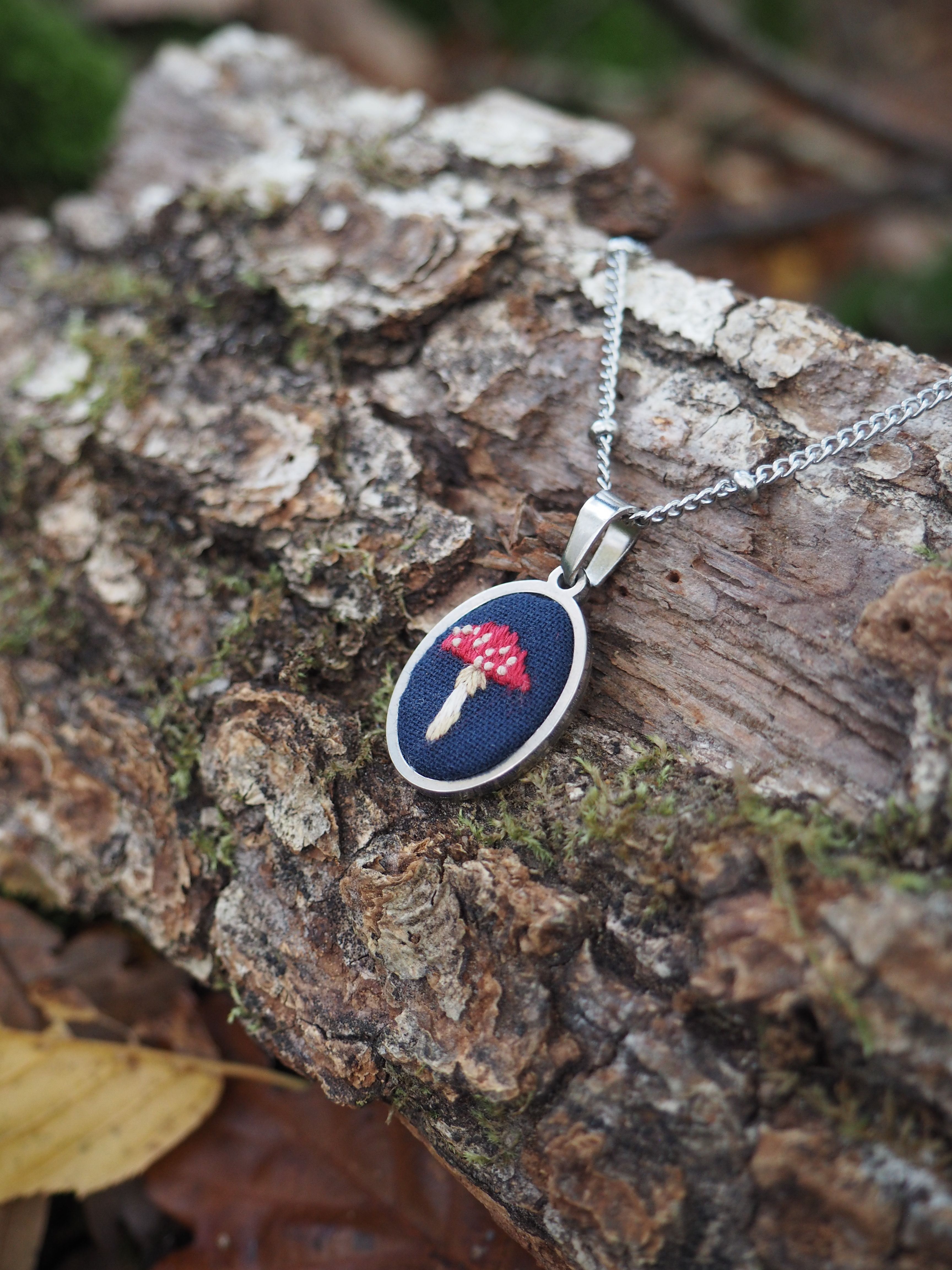 Cette image présente un bijou délicat posé sur un tronc d'arbre dans un environnement naturel. Il s'agit d'un pendentif ovale monté sur une chaîne argentée fine. Le pendentif contient ce qui semble être une véritable fleur séchée ou pressée - probablement une petite fleur rose ou rouge - préservée dans une résine transparente ou sous verre, créant un effet de capsule temporelle botanique. Le bijou repose sur l'écorce rugueuse et texturée d'un tronc d'arbre, avec de la mousse visible qui ajoute une touche de vert naturel à la composition. L'arrière-plan est artistiquement flouté, créant un effet de profondeur de champ qui met en valeur le pendentif comme sujet principal. Cette photographie capture harmonieusement le contraste entre l'objet manufacturé délicat et l'environnement naturel brut, suggérant une connexion poétique entre la nature préservée dans le bijou et la nature vivante qui l'entoure. L'éclairage naturel doux met en valeur les détails du pendentif tout en créant une atmosphère sereine et contemplative.