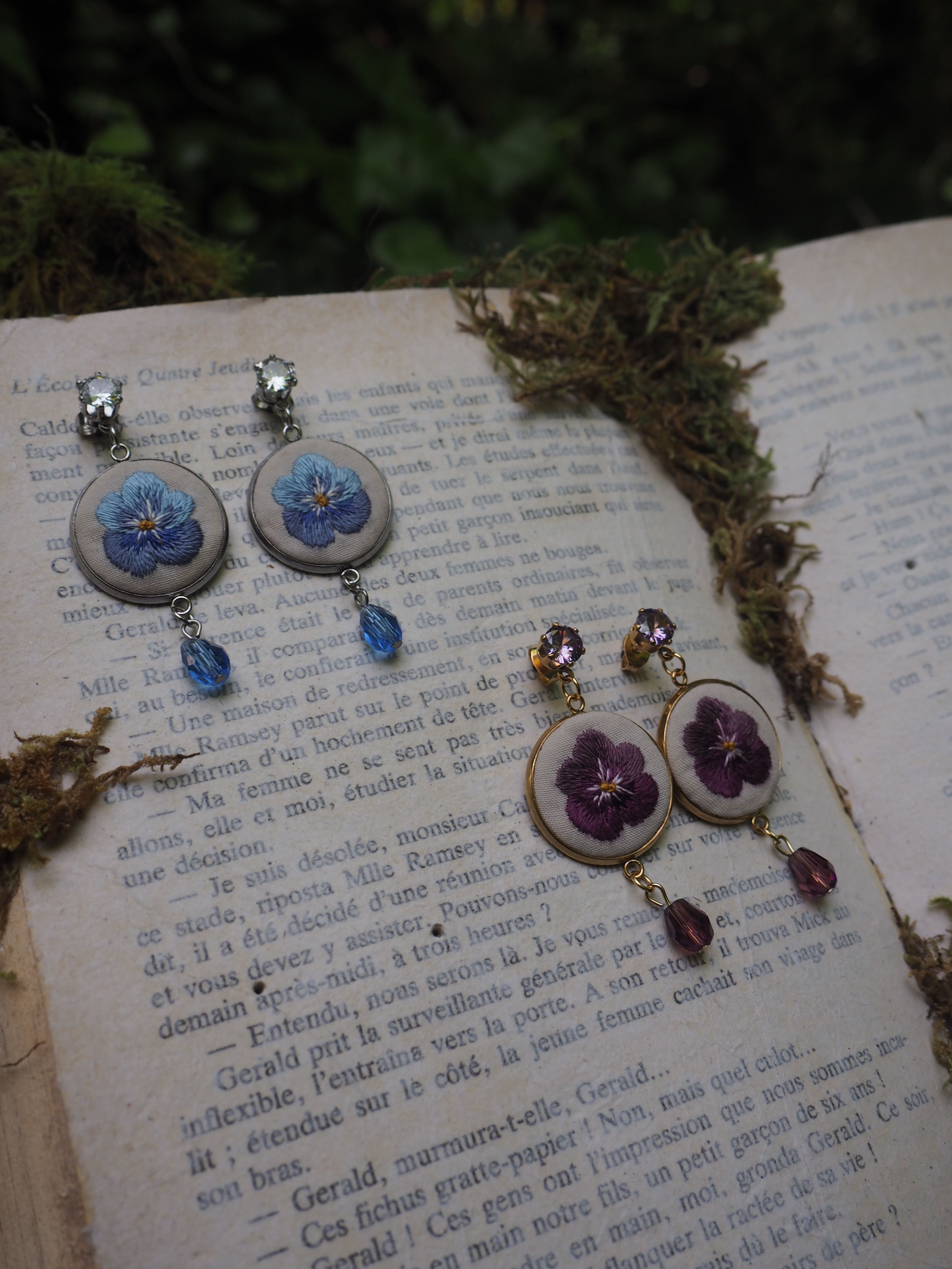 Cette image montre de jolies boucles d'oreilles artisanales posées sur les pages d'un livre ancien. Il y a deux paires de boucles d'oreilles : une paire avec des fleurs bleues et une autre avec des fleurs violettes. Les boucles d'oreilles semblent être faites dans un style vintage ou romantique, avec des pendentifs circulaires contenant les motifs floraux et des petites perles ou cristaux qui pendent en dessous. L'arrière-plan montre du texte imprimé sur les pages du livre et ce qui semble être de la mousse ou de la verdure sur le côté. La composition crée une ambiance très poétique et romantique, parfaite pour mettre en valeur ces bijoux délicats.