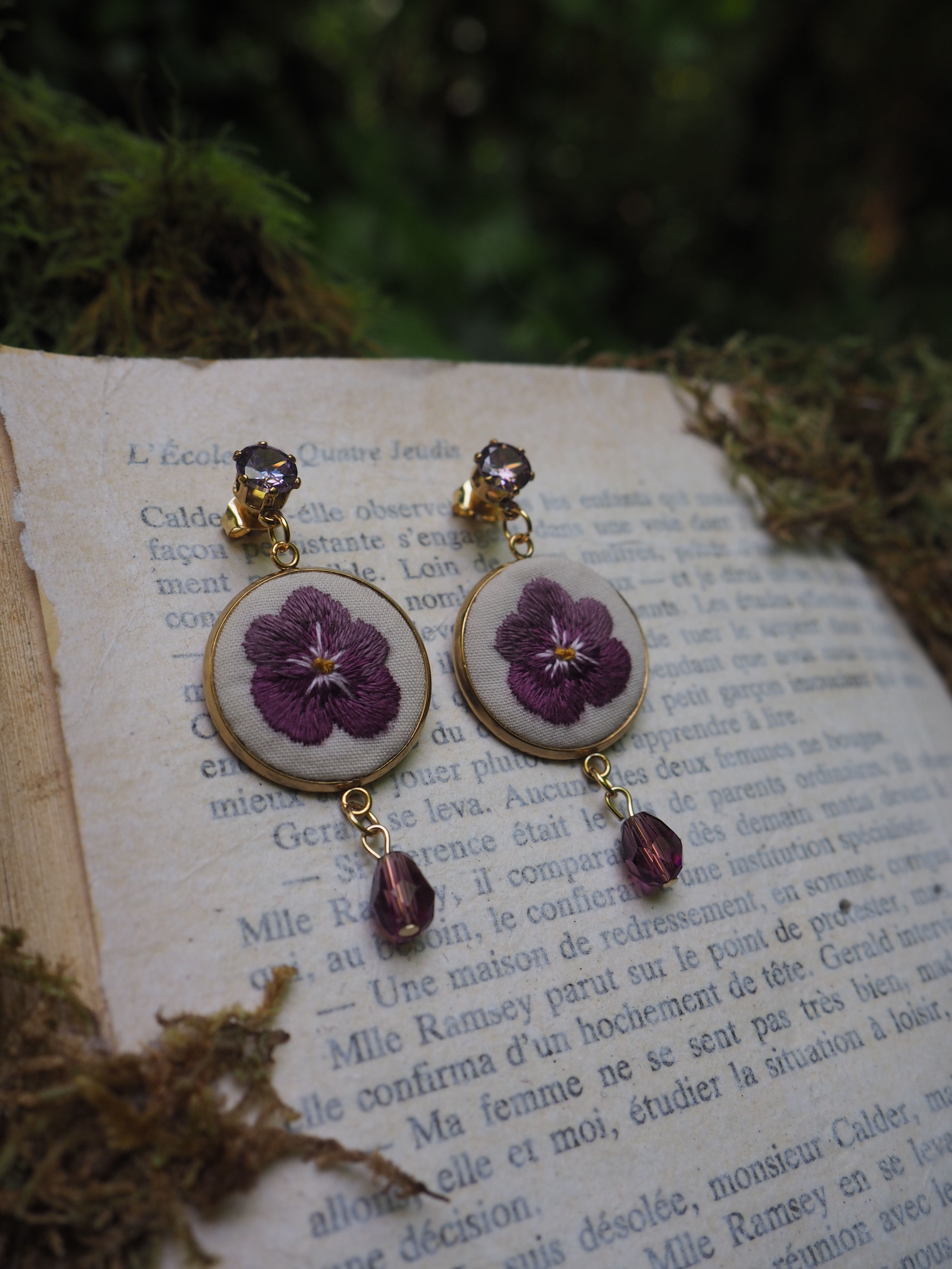 Ces boucles d'oreilles sont vraiment charmantes! Elles présentent de véritables fleurs pressées violettes (qui semblent être des pensées) préservées dans ce qui paraît être des supports circulaires blancs. Les boucles sont agrémentées de petites perles violettes qui pendent en dessous, créant un effet délicat et romantique. L'image est mise en scène sur ce qui semble être une page de livre en français, donnant une touche très poétique et vintage à la composition. Les tons violets des fleurs contrastent magnifiquement avec le fond blanc du support et la teinte jaunâtre de la page de livre. C'est un excellent exemple de bijoux artisanaux qui incorporent des éléments naturels de manière élégante.