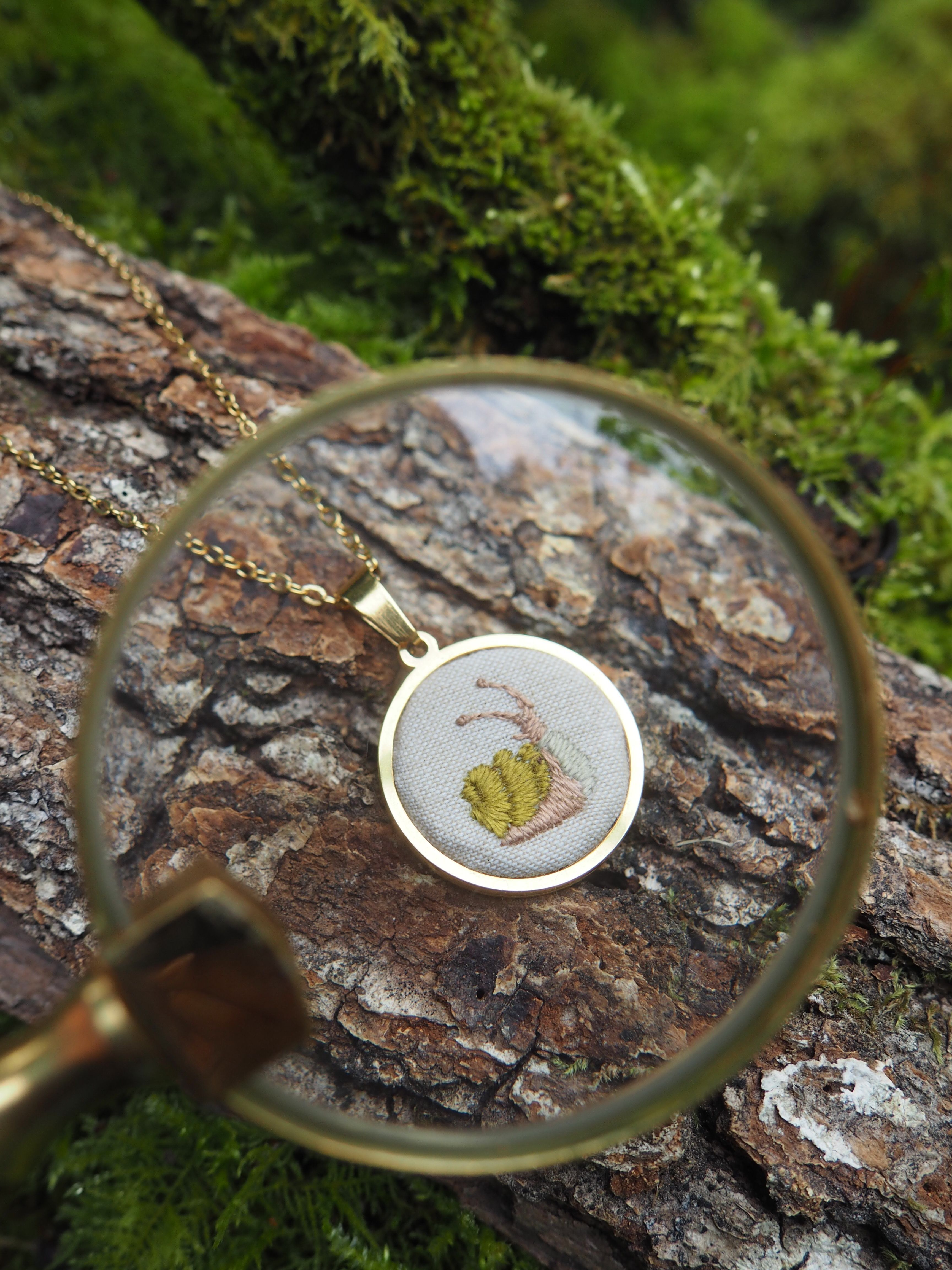 Cette image présente une composition artistique et détaillée, centrée sur un pendentif délicat posé sur une surface naturelle. Au premier plan, une loupe dorée est positionnée de manière à encadrer le pendentif, créant un effet de mise au point et d'agrandissement. Le bord de la loupe est visible, avec son cadre métallique brillant. À travers la loupe, on peut voir un pendentif rond, également avec un cadre doré. À l'intérieur de ce pendentif, il y a une petite broderie ou illustration sur un fond clair, représentant ce qui semble être une petite plante ou un élément végétal stylisé, avec des tons de vert et de beige ou de marron clair. Le pendentif est posé sur un morceau d'écorce d'arbre texturée, de couleur marron foncé avec des nuances plus claires et des creux. L'écorce est recouverte par endroits de mousse verte, ajoutant une touche de nature et de couleur. L'arrière-plan est flou, mais on distingue davantage de mousse verte luxuriante et d'autres éléments végétaux, suggérant un environnement forestier ou naturel. La lumière semble douce et naturelle, mettant en valeur les textures et les couleurs des différents éléments. L'ensemble de l'image évoque la minutie, l'artisanat et un lien avec la nature, avec la loupe invitant le spectateur à observer les détails.