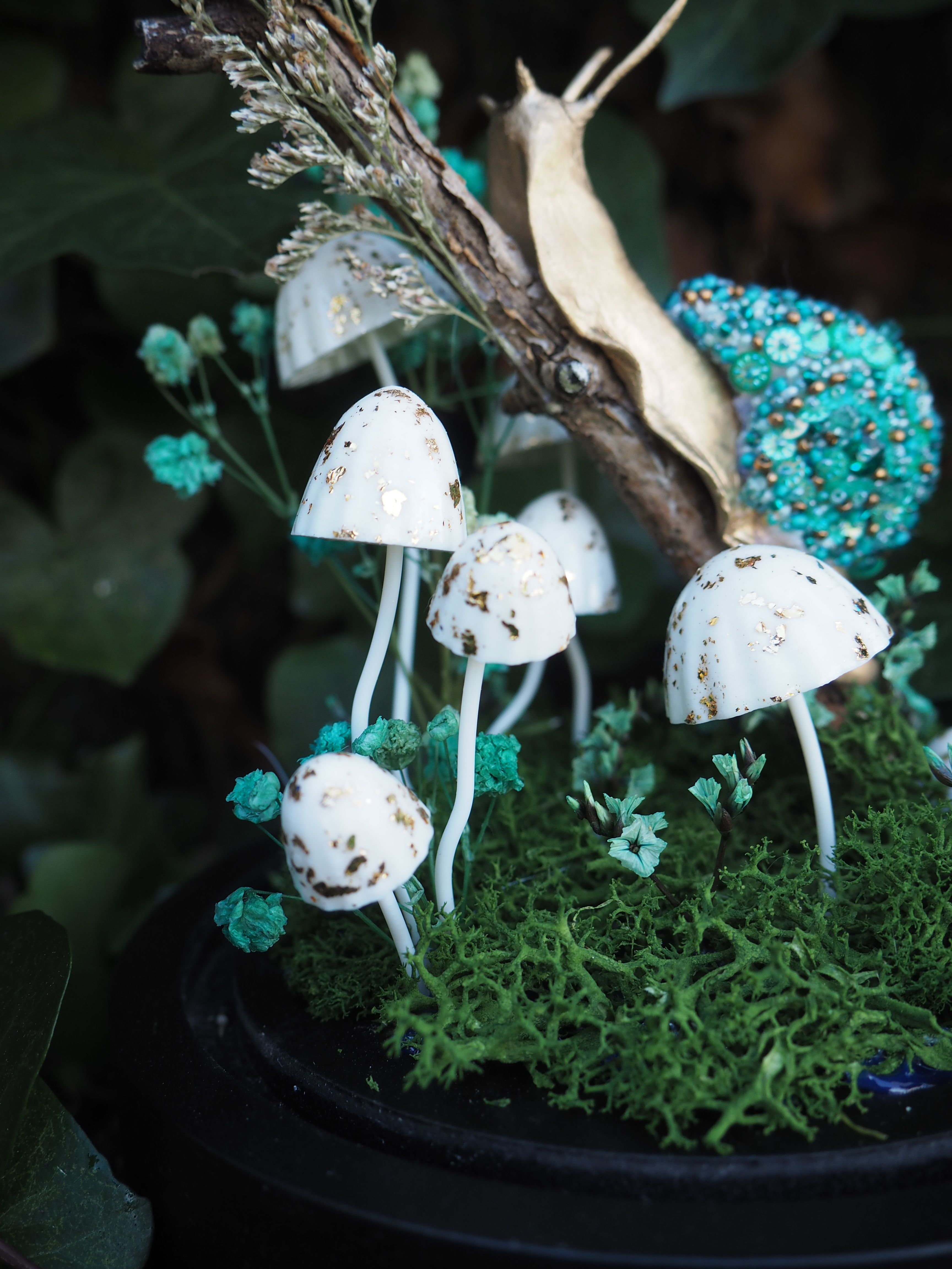 Cette image présente une scène miniature enchantée, centrée sur un arrangement de champignons et d'un escargot dans un environnement de mousse et de verdure. Au premier plan, on voit plusieurs petits champignons blancs avec des taches dorées, qui semblent être des décorations. Ils sont de tailles variées et sont plantés dans un lit de mousse verte texturée, qui occupe la majeure partie du bas de l'image. Entre les champignons et la mousse, on aperçoit de petites touffes de verdure d'un vert plus clair, presque turquoise, ajoutant de la profondeur et de la couleur à la scène. Au-dessus des champignons, un escargot stylisé est posé sur une branche ou un morceau de bois. La coquille de l'escargot est d'un bleu-vert vif, ornée de petites perles ou de paillettes qui lui donnent un aspect scintillant et texturé. Le corps de l'escargot est d'une couleur dorée ou beige clair, contrastant avec sa coquille. L'arrière-plan est flou, suggérant une profondeur de champ faible, ce qui met l'accent sur les éléments du premier plan. On distingue des teintes sombres de vert, probablement des feuilles ou de la végétation, qui contribuent à l'ambiance mystérieuse et forestière de la scène. L'ensemble de l'image dégage une atmosphère féerique et délicate, comme une petite scène de conte de fées ou un terrarium décoratif.