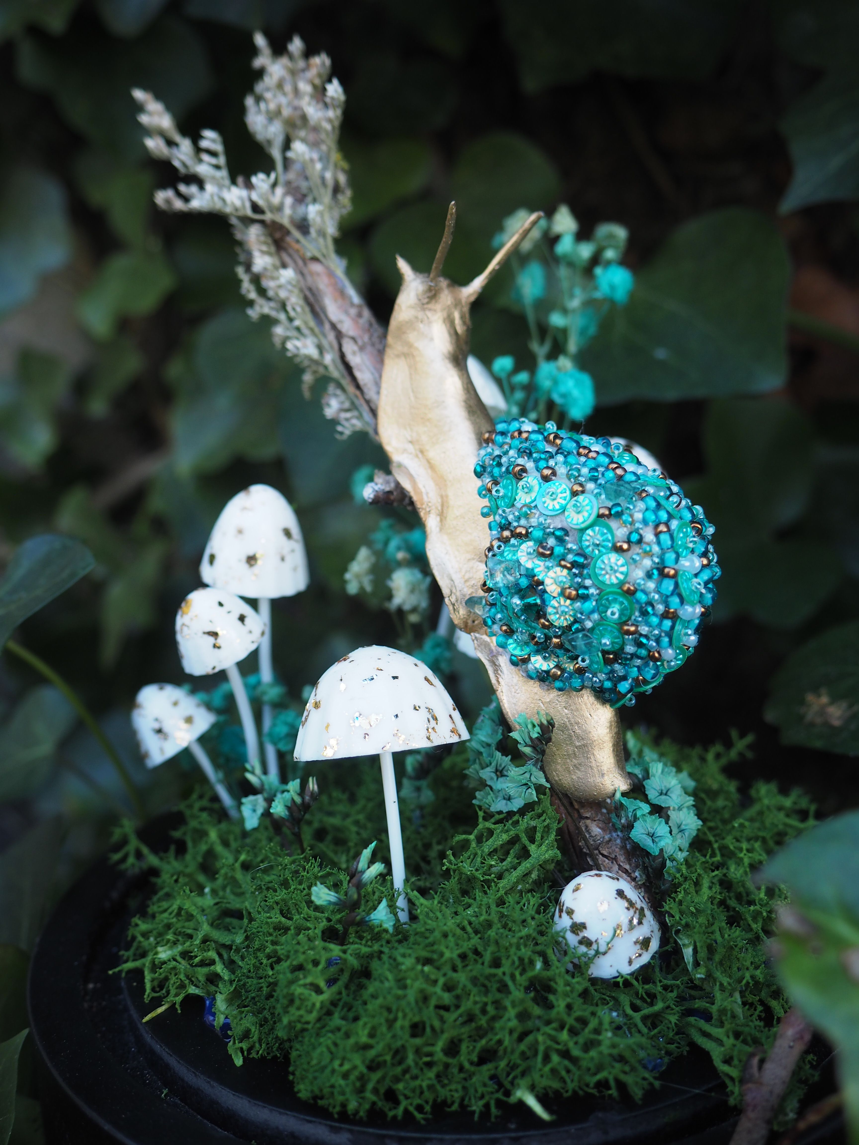 Cette image présente une composition artistique et fantaisiste, centrée sur une figurine d'escargot ornée, placée dans un décor miniature de forêt. Au premier plan, on voit un escargot stylisé, dont le corps est d'une couleur dorée ou laiton, lui donnant un aspect précieux. Sa coquille est particulièrement remarquable, recouverte d'une multitude de perles et de petites billes de couleur turquoise et bleu-vert, créant une texture riche et scintillante. L'escargot est positionné sur une branche ou un morceau de bois sec, qui semble être son support principal. Autour de l'escargot et à sa base, un tapis de mousse verte, dense et texturée, évoque un sous-bois. Plusieurs petits champignons miniatures, de couleur blanche avec des taches sombres (peut-être noires ou marron foncé), sont dispersés sur la mousse, ajoutant une touche de réalisme et de conte de fées à la scène. Certains de ces champignons ont des formes variées, certains étant plus élancés, d'autres plus trapus. En arrière-plan, on distingue un feuillage vert foncé et flou, suggérant un environnement naturel et luxuriant, comme un jardin ou une forêt. Quelques petites fleurs ou éléments végétaux de couleur bleu-vert pâle sont également visibles, ajoutant de la profondeur et de la couleur à l'ensemble. L'éclairage semble doux et naturel, mettant en valeur les détails et les textures des différents éléments. L'ensemble de la composition est probablement une œuvre d'art décorative ou un diorama, conçu pour évoquer un monde miniature enchanté.