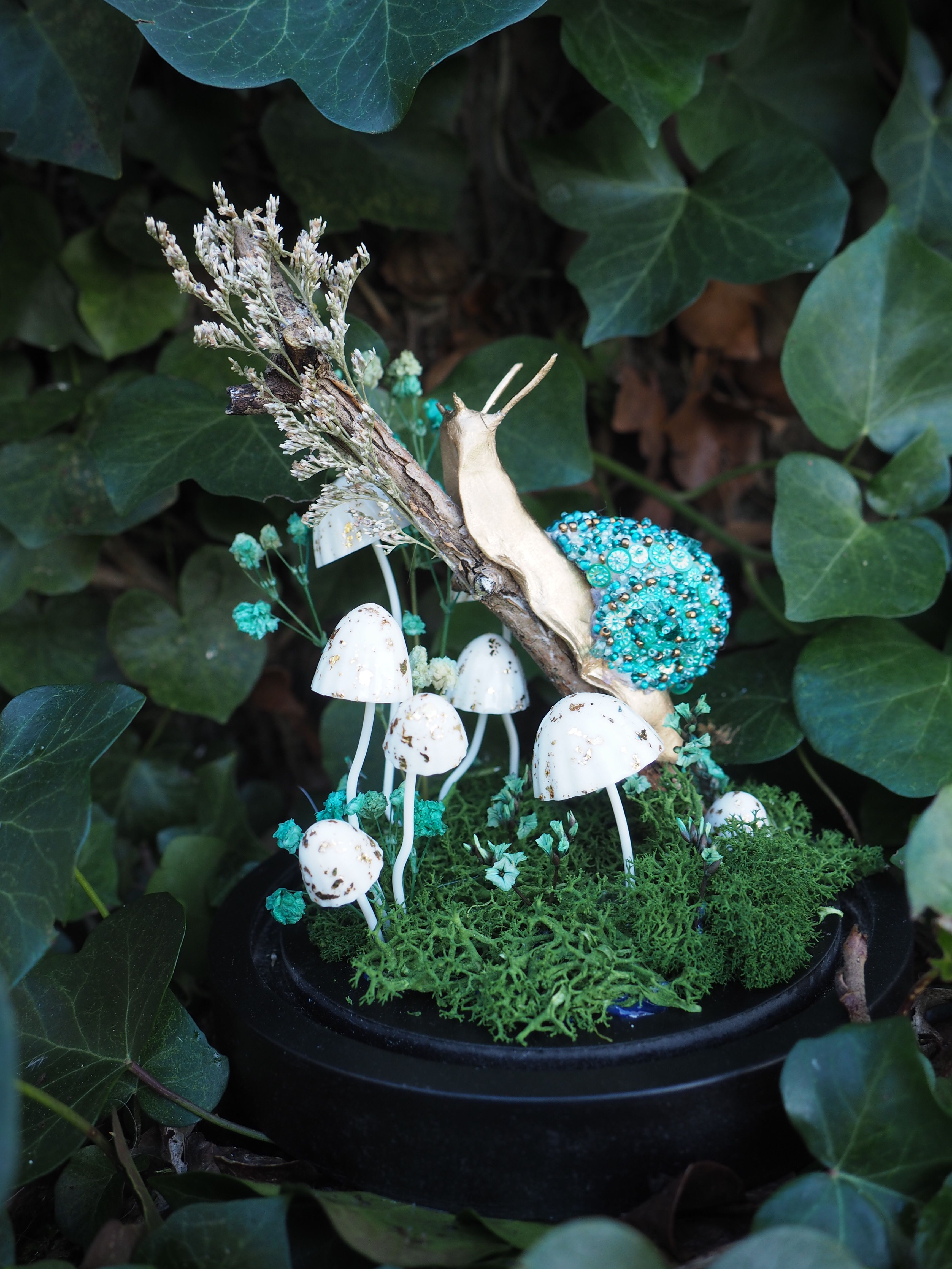 L'image présente une composition artistique et fantaisiste, évoquant un petit écosystème miniature ou un terrarium, avec un thème forestier et féerique. Au centre de l'image, un escargot doré, stylisé et brillant, est positionné sur une branche ou un morceau de bois sec. Sa coquille est ornée de petites perles ou de paillettes bleues-vertes scintillantes, lui donnant un aspect précieux et irréel. Sous l'escargot et la branche, un groupe de petits champignons blancs, aux chapeaux ronds et aux pieds fins, pousse sur un lit de mousse verte luxuriante. Certains champignons sont plus grands et bien visibles, tandis que d'autres sont plus petits et se fondent davantage dans la mousse. La mousse verte forme un tapis dense et texturé, créant un sol naturel pour cette scène miniature. Quelques petites fleurs ou éléments décoratifs de couleur bleu-vert clair sont parsemés dans la mousse et autour des champignons, ajoutant une touche de couleur et de délicatesse. L'ensemble de cette composition est contenu dans un récipient sombre, probablement un pot ou une coupelle noire, qui sert de base et de contraste aux couleurs vives de la scène. L'arrière-plan est flou et dominé par des feuilles de lierre vert foncé, créant une ambiance naturelle et mystérieuse, comme si cette scène miniature était cachée au cœur d'une forêt. La lumière semble douce et diffuse, mettant en valeur les détails et les textures de chaque élément. L'image dégage une atmosphère de conte de fées, de magie et de nature enchantée, avec une attention particulière aux détails et à l'esthétique.