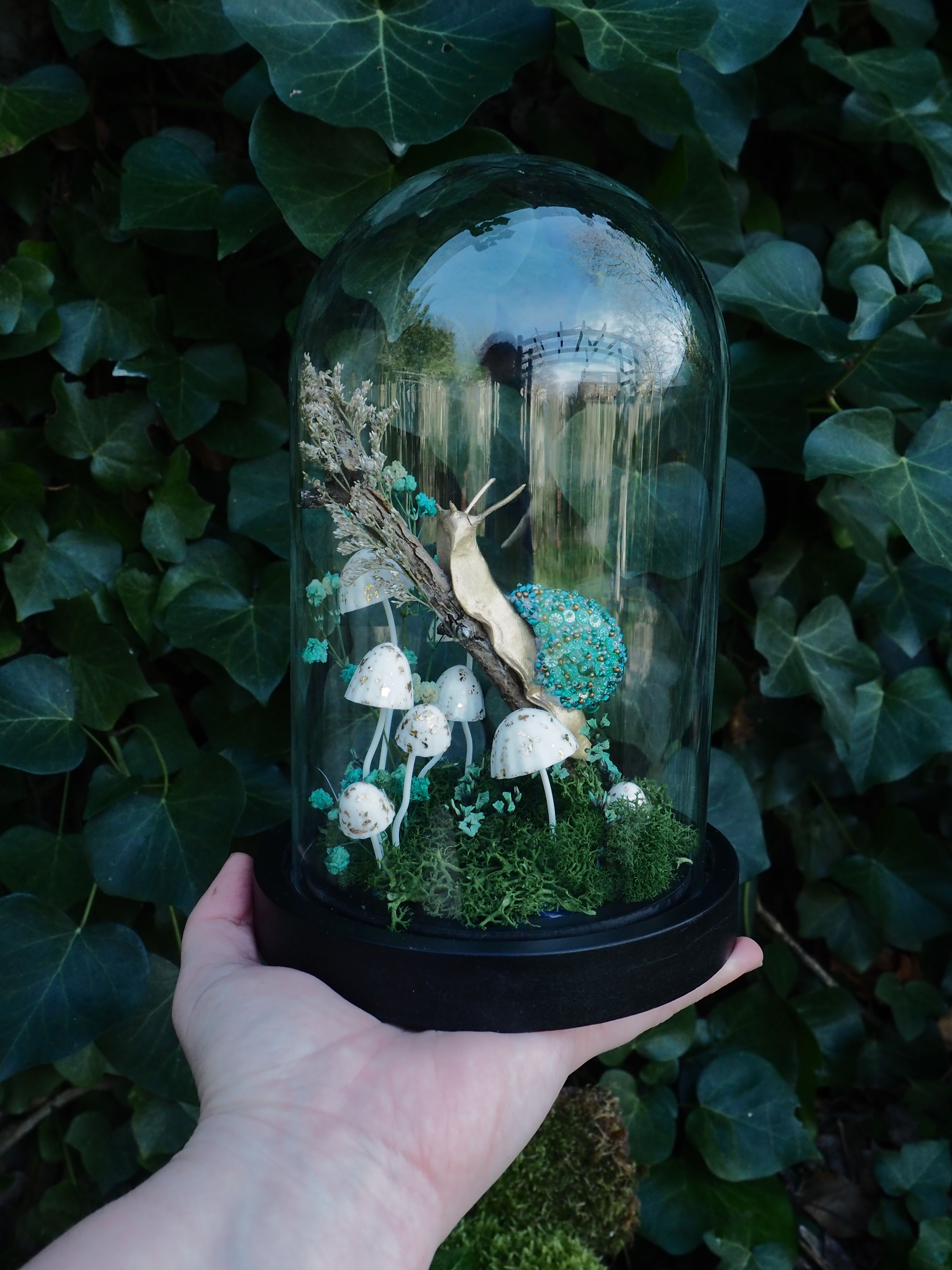This image features a close-up shot of a person's hand holding a glass cloche, which encases a miniature, whimsical diorama. The cloche has a black base and a clear, domed glass cover. Inside the cloche, a fantastical scene unfolds:
*   **Central Figure:** A prominent, elongated snail-like creature with a light, almost golden-brown body and a vibrant, textured, teal-colored shell is positioned towards the upper center. It appears to be climbing or resting on a piece of dried, light-colored foliage or wood.
*   **Mushrooms:** Several small, white-capped mushrooms with slender stems are scattered across the base of the diorama. They vary slightly in size and arrangement, adding to the enchanted forest aesthetic.
*   **Foliage/Moss:** The base of the diorama is covered with lush, green moss, creating a natural ground cover. There are also some dried, lighter-colored plant elements, possibly twigs or dried flowers, contributing to the texture and depth of the scene.
*   **Background Elements:** In the background within the cloche, through the glass, there are hints of what appears to be a miniature bridge or structure, suggesting a larger, imagined landscape. The hand holding the cloche is light-skinned, with visible fingers and palm, providing a sense of scale to the miniature artwork. The background outside the cloche is dominated by dark green ivy leaves, which are slightly out of focus, emphasizing the cloche and its contents. The lighting appears natural, highlighting the details within the glass dome. Overall, the image conveys a sense of delicate craftsmanship and a magical, miniature world contained within the glass.