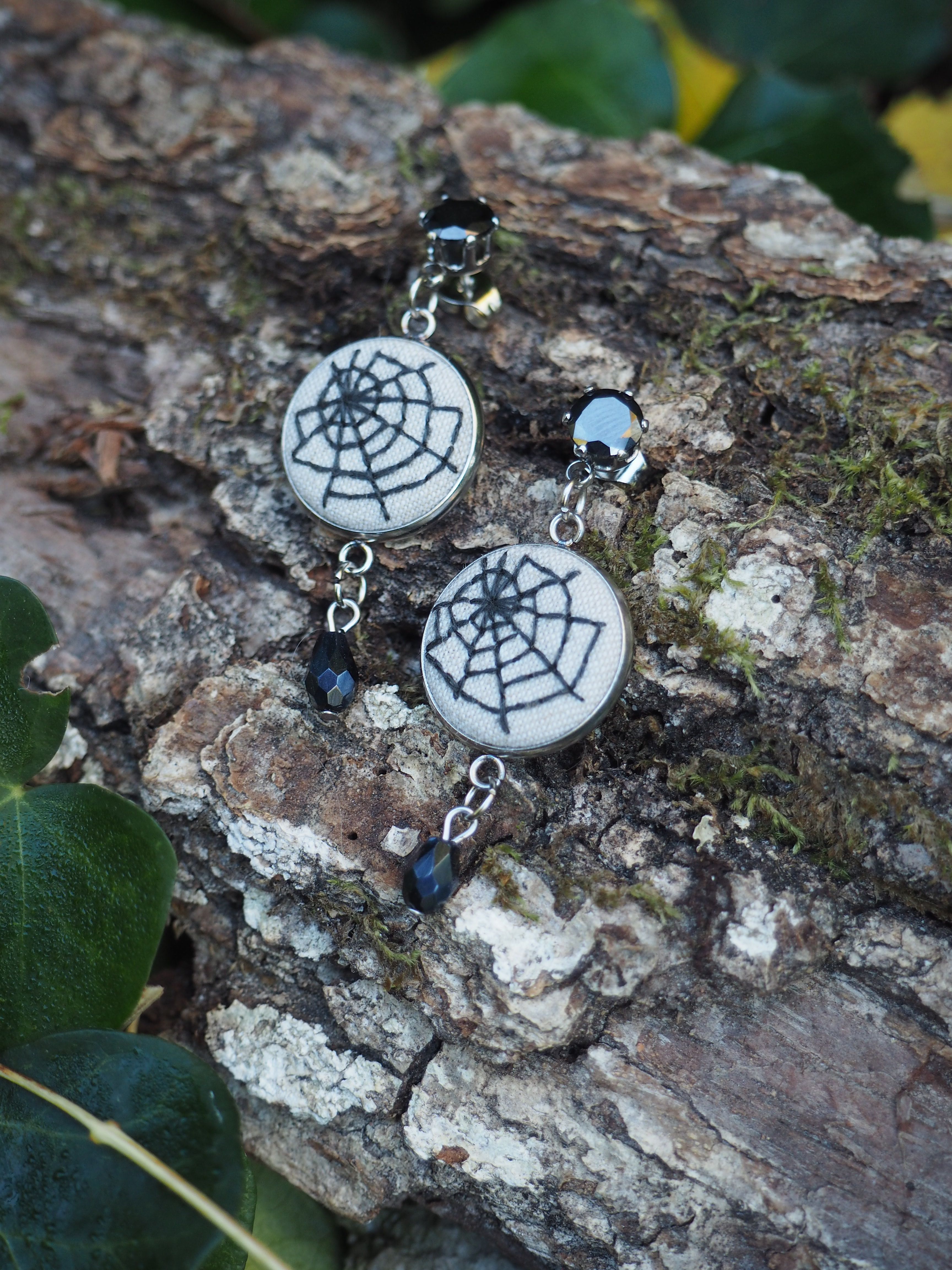 Cette image présente une paire de boucles d'oreilles artisanales disposées sur un morceau d'écorce d'arbre texturée. Les bijoux sont composés de pendentifs circulaires blancs ornés d'un motif de toile d'araignée noire au centre. Chaque boucle d'oreille est agrémentée de petites perles noires qui ajoutent une touche d'élégance au design. L'écorce qui sert de support présente une surface rugueuse et naturelle avec des tons bruns et gris, créant un contraste intéressant avec les bijoux. On peut apercevoir quelques feuilles vertes en arrière-plan, ce qui renforce l'aspect naturel de la composition. Le style des boucles d'oreilles évoque un thème gothique ou mystique avec le motif de toile d'araignée, tout en conservant une esthétique moderne et raffinée grâce au contraste noir et blanc et à la finition soignée des éléments.