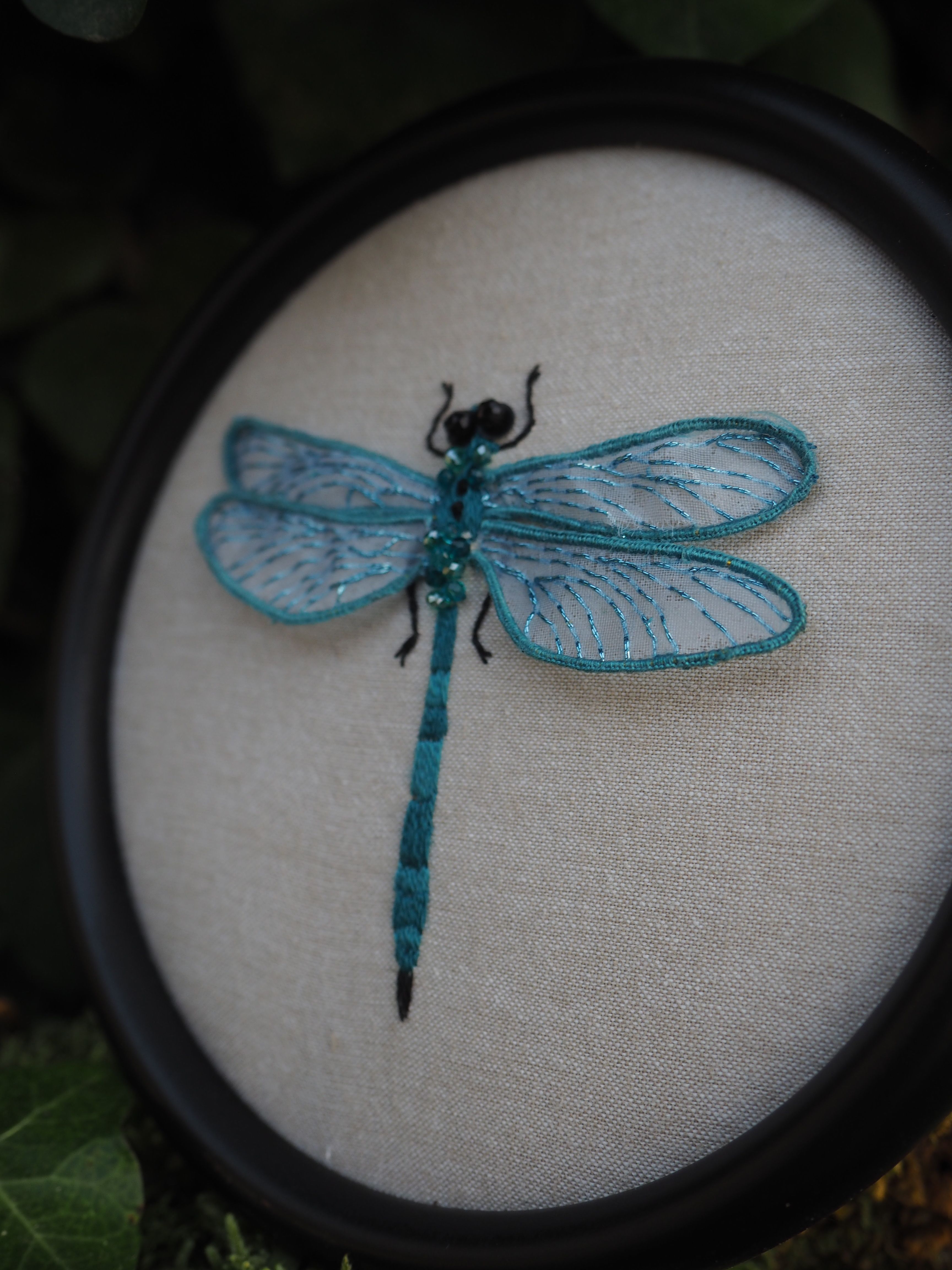 L'image présente une broderie délicate d'une libellule bleue, encadrée dans un cercle noir. La libellule est le sujet central et est représentée avec des détails fins. **Description de la libellule :**
*   **Couleur :** La libellule est principalement de couleur bleu-vert turquoise, avec des nuances plus claires et plus foncées qui lui donnent de la profondeur.
*   **Corps :** Le corps est long et segmenté, typique des libellules, avec une tête plus foncée et des yeux noirs.
*   **Ailes :** Les ailes sont translucides et d'un bleu plus clair, avec des nervures brodées en bleu foncé qui imitent la structure naturelle des ailes d'insectes. On peut percevoir un léger scintillement ou une texture irisée sur les ailes, suggérant l'utilisation de fils spéciaux ou de perles fines.
*   **Texture :** La broderie semble avoir une texture en relief, ce qui ajoute à son réalisme et à son attrait visuel. **Description du cadre et du fond :**
*   **Cadre :** La broderie est montée dans un cadre circulaire noir, qui contraste bien avec les couleurs claires de la libellule et du fond.
*   **Fond :** Le fond sur lequel la libellule est brodée est un tissu de couleur crème ou blanc cassé, probablement du lin ou un tissu similaire, qui met en valeur la broderie sans la surcharger.
*   **Profondeur :** L'image est prise avec une faible profondeur de champ, ce qui rend la libellule nette et le fond légèrement flou, attirant l'attention sur l'œuvre d'art. **Contexte de l'image :**
*   **Arrière-plan :** L'arrière-plan de la photo est flou et semble être composé de feuillage vert foncé, peut-être des feuilles de lierre ou d'autres plantes, ce qui crée une ambiance naturelle et met en scène la libellule comme si elle était dans son environnement naturel.
*   **Lumière :** La lumière semble naturelle et douce, éclairant la broderie de manière à faire ressortir les couleurs et les textures. Dans l'ensemble, l'image est une belle représentation d'une œuvre d'art artisanale, mettant en valeur la finesse et la beauté de la broderie d'une libellule.
