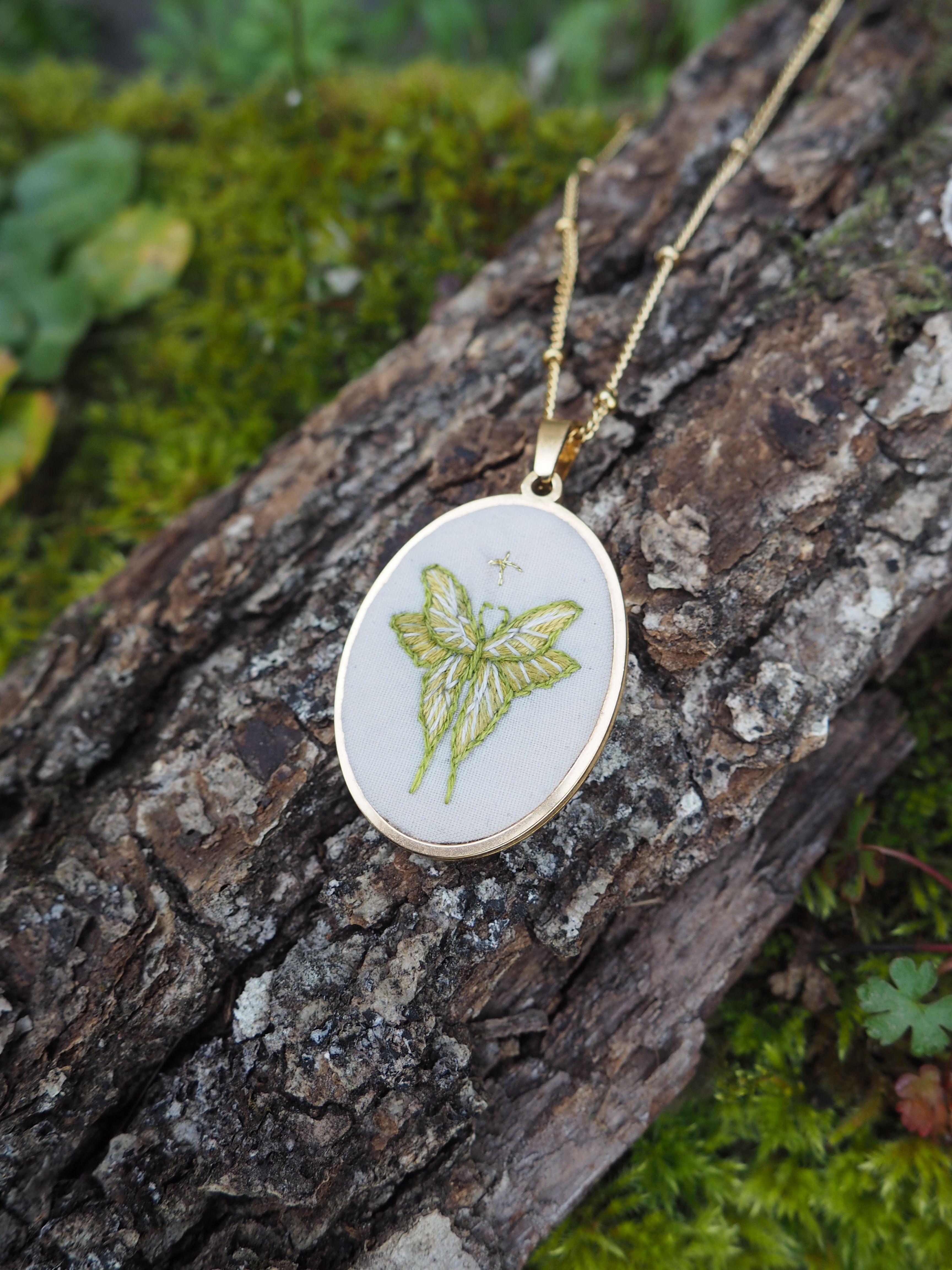 Cette image montre un pendentif délicat sur une chaîne dorée, photographié sur un morceau de bois couvert de mousse. Le pendentif est rond et présente une broderie ou un dessin d'une libellule en vert clair sur un fond blanc. Le design est élégant et naturel, s'harmonisant bien avec l'arrière-plan naturel de l'écorce texturée et de la mousse verte. C'est un bel exemple de bijou inspiré par la nature, combinant la délicatesse de la broderie avec un motif d'insecte gracieux.