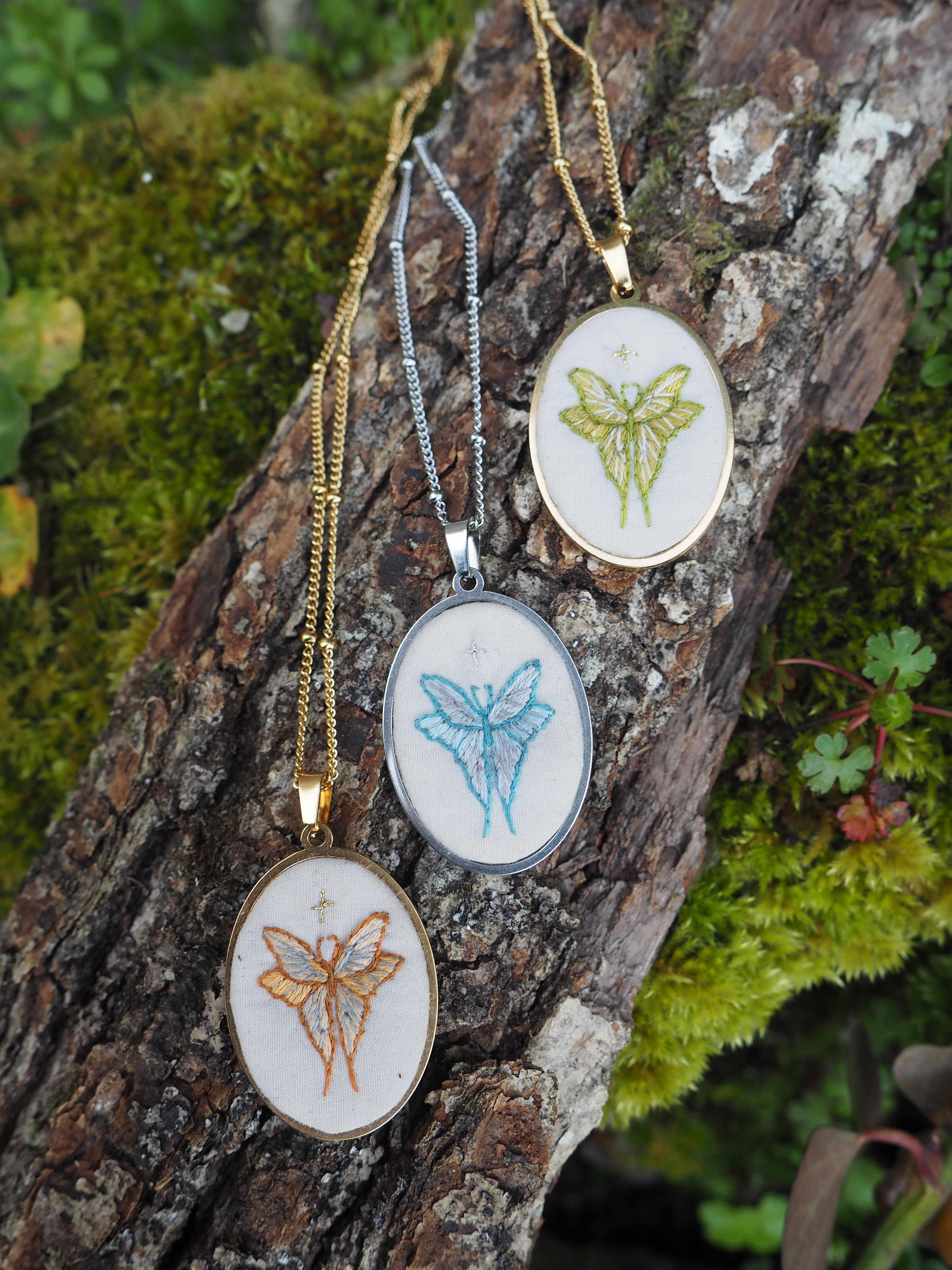 Cette image montre trois jolis pendentifs brodés disposés sur un morceau d'écorce couvert de mousse. Chaque pendentif présente un papillon brodé dans des couleurs différentes - l'un en jaune/vert, un autre en bleu clair, et le troisième en orange/doré. Les pendentifs sont montés sur des chaînes de différentes couleurs assorties (dorée et argentée) et semblent être des pièces de bijouterie artisanale délicate. Le cadre naturel avec l'écorce moussue et la verdure en arrière-plan met parfaitement en valeur ces créations délicates. C'est une belle composition qui marie l'artisanat et la nature.