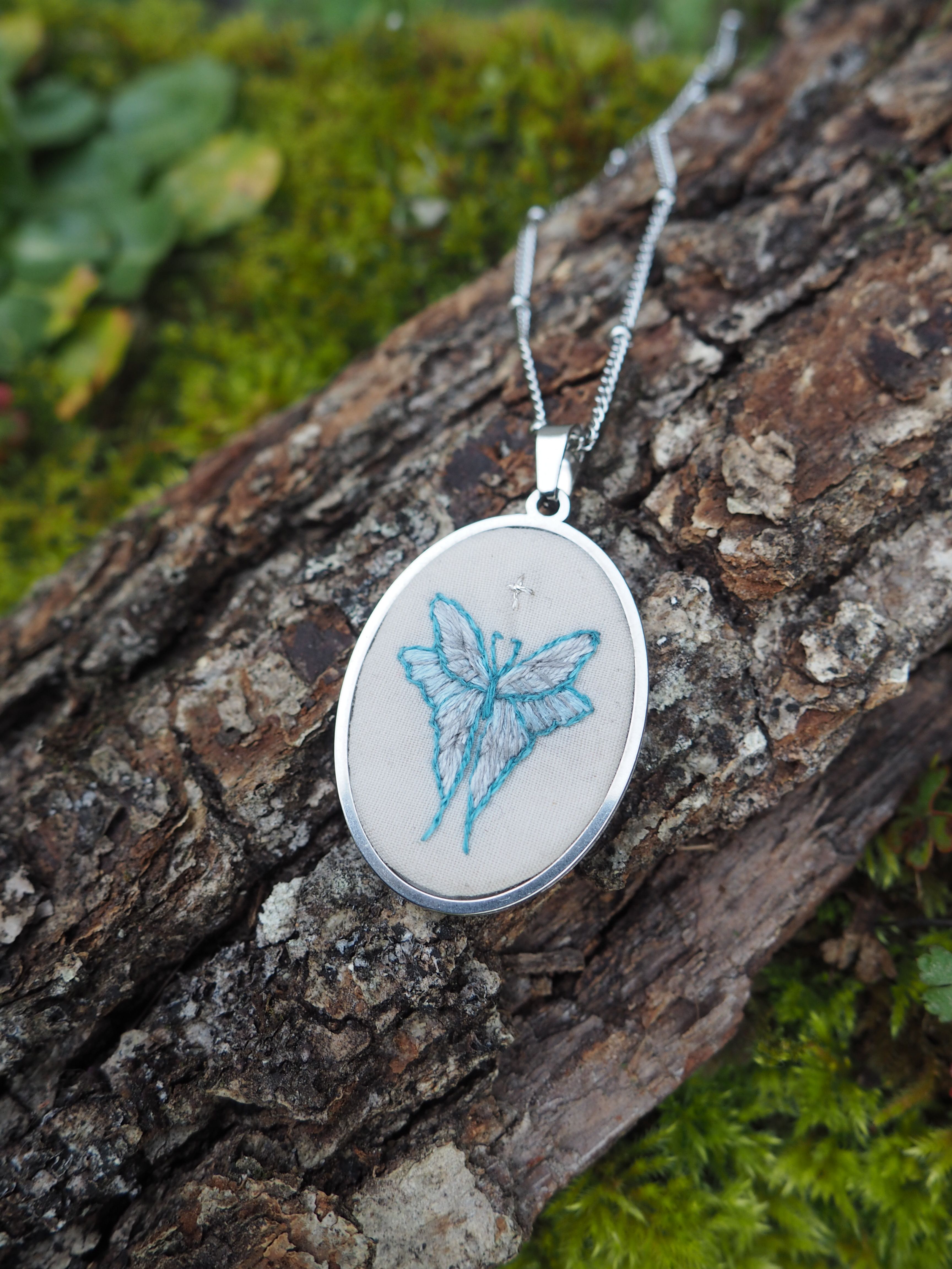 Cette image montre un pendentif délicat sur une chaîne en argent, photographié sur un morceau d'écorce d'arbre couvert de mousse verte. Le pendentif est rond et contient une broderie ou un dessin d'un papillon bleu sur un fond blanc. Le design est simple mais élégant, avec le papillon représenté dans des tons bleu clair. Le contraste entre le bijou argenté et la texture naturelle rugueuse de l'écorce, ainsi que la mousse verte, crée une belle composition photographique qui met en valeur la délicatesse du pendentif.