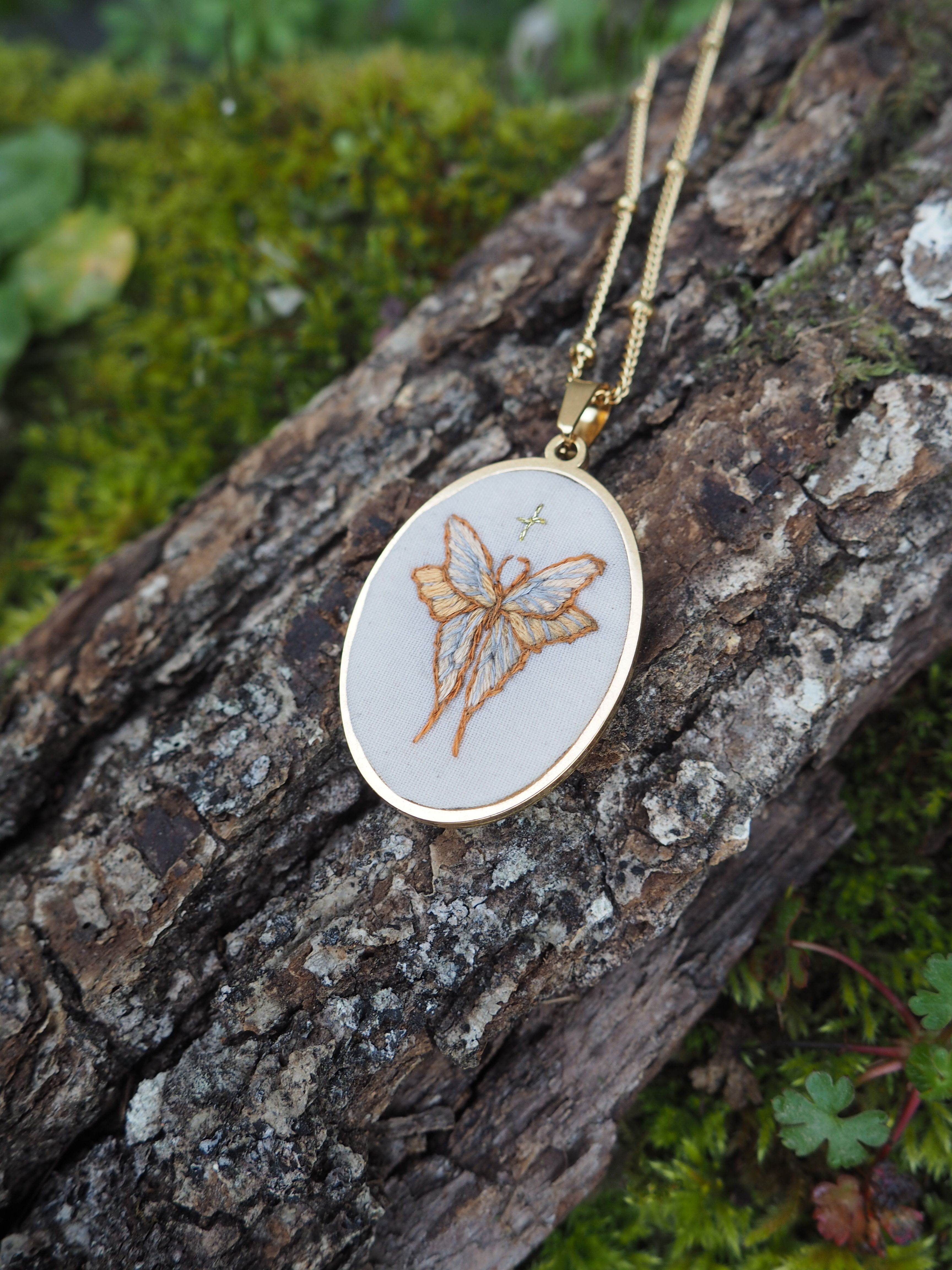Cette image montre un délicat pendentif artisanal composé d'un cercle à broder (tambour de broderie miniature) monté en collier avec une chaîne dorée. Le motif brodé représente un papillon dans des tons dorés ou beiges sur un fond blanc. Le pendentif est photographié sur un tronc d'arbre recouvert de mousse verte, ce qui crée un beau contraste naturel avec le bijou. C'est une pièce de bijouterie très délicate et naturelle qui allie l'art de la broderie à la joaillerie.