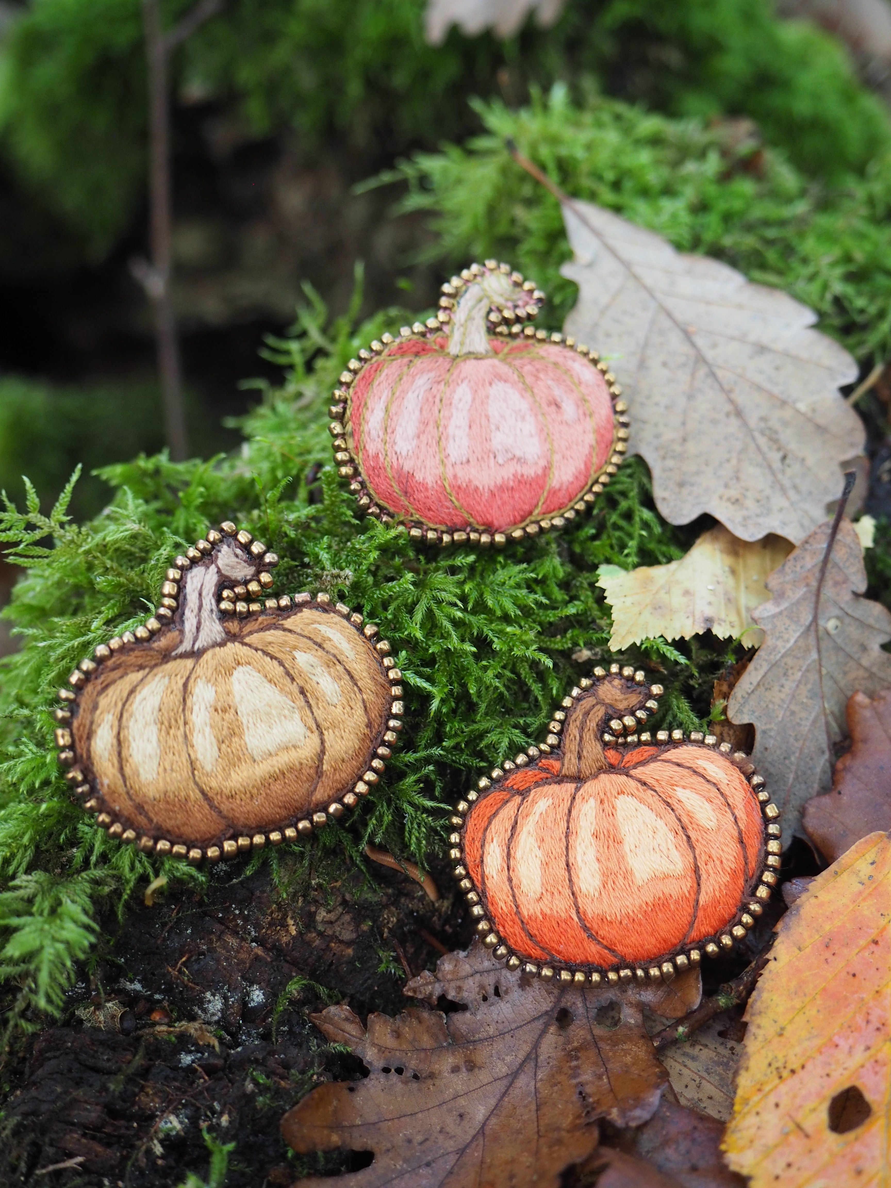 Cette image présente une composition automnale très artistique et décorative. On y voit trois pendentifs ou ornements en forme de citrouilles, disposés harmonieusement sur un lit de mousse verte luxuriante. **Les éléments principaux :** - **Trois citrouilles décoratives** aux teintes variées : une orange vif, une plus claire tirant vers le beige/crème, et une troisième dans des tons orangés
- Chaque citrouille est ornée d'un **contour perlé doré** qui leur donne un aspect précieux et raffiné
- Elles sont équipées de **petites chaînes ou attaches** suggérant qu'il s'agit de bijoux ou d'ornements décoratifs **L'environnement :**
- Un **tapis de mousse** d'un vert éclatant qui sert d'écrin naturel
- Des **feuilles d'automne** aux couleurs chaudes (brun, ocre) qui complètent parfaitement la palette chromatique
- L'ensemble évoque une **ambiance forestière** et saisonnière Cette mise en scène capture parfaitement l'esprit de l'automne, mêlant éléments naturels et créations artisanales dans une harmonie de couleurs et de textures très réussie.