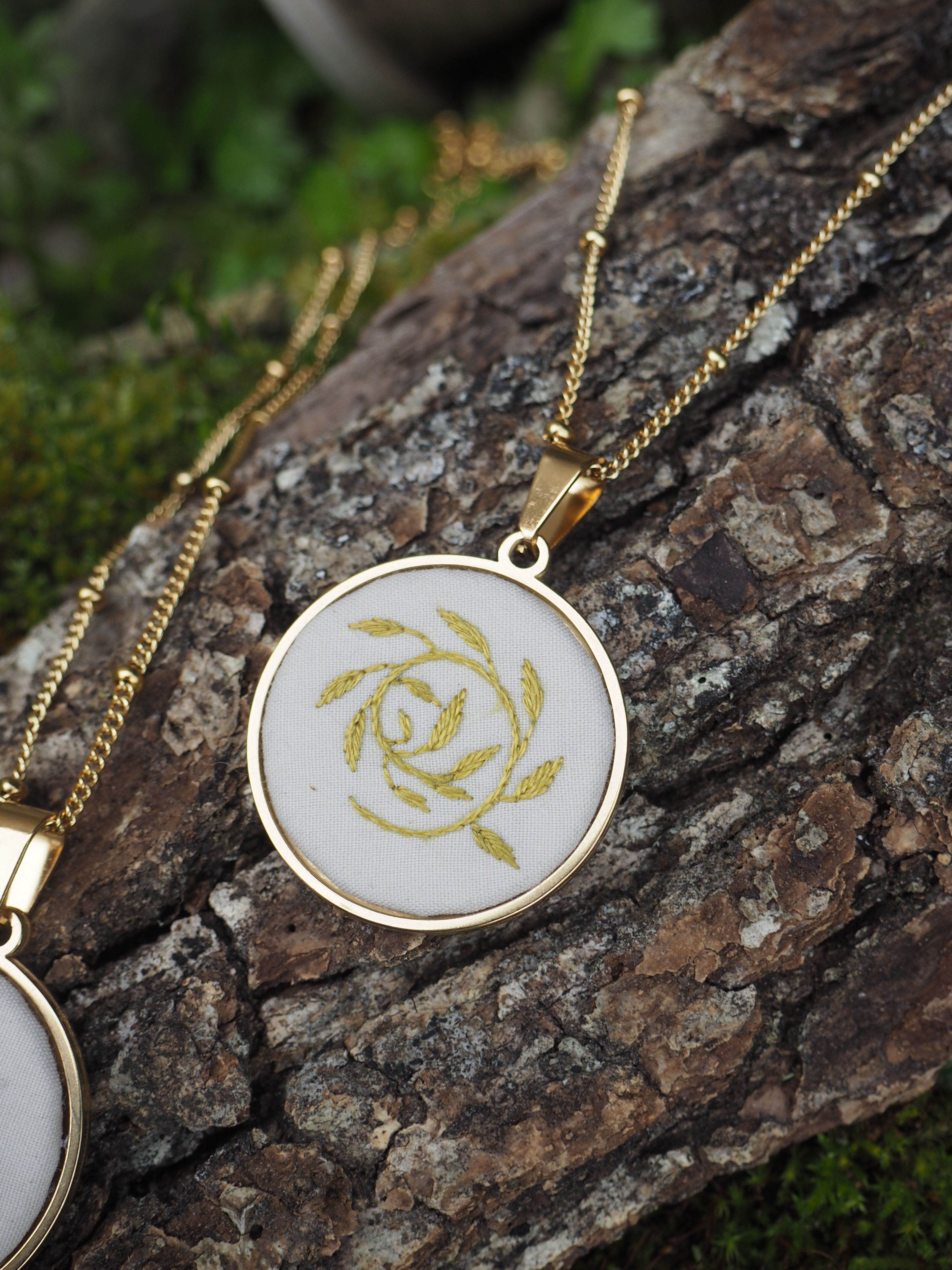 Cette image montre un pendentif délicat sur une chaîne dorée, posé sur un morceau de bois couvert de mousse. Le pendentif est de forme ovale avec un cadre doré et contient ce qui semble être une broderie ou un motif de rose stylisée en fil doré sur un fond blanc ou crème. La photographie est prise en gros plan avec une belle profondeur de champ, mettant en valeur les détails du bijou tout en montrant la texture naturelle du bois et la mousse verte en arrière-plan. C'est une composition élégante qui marie les éléments naturels avec un bijou raffiné.