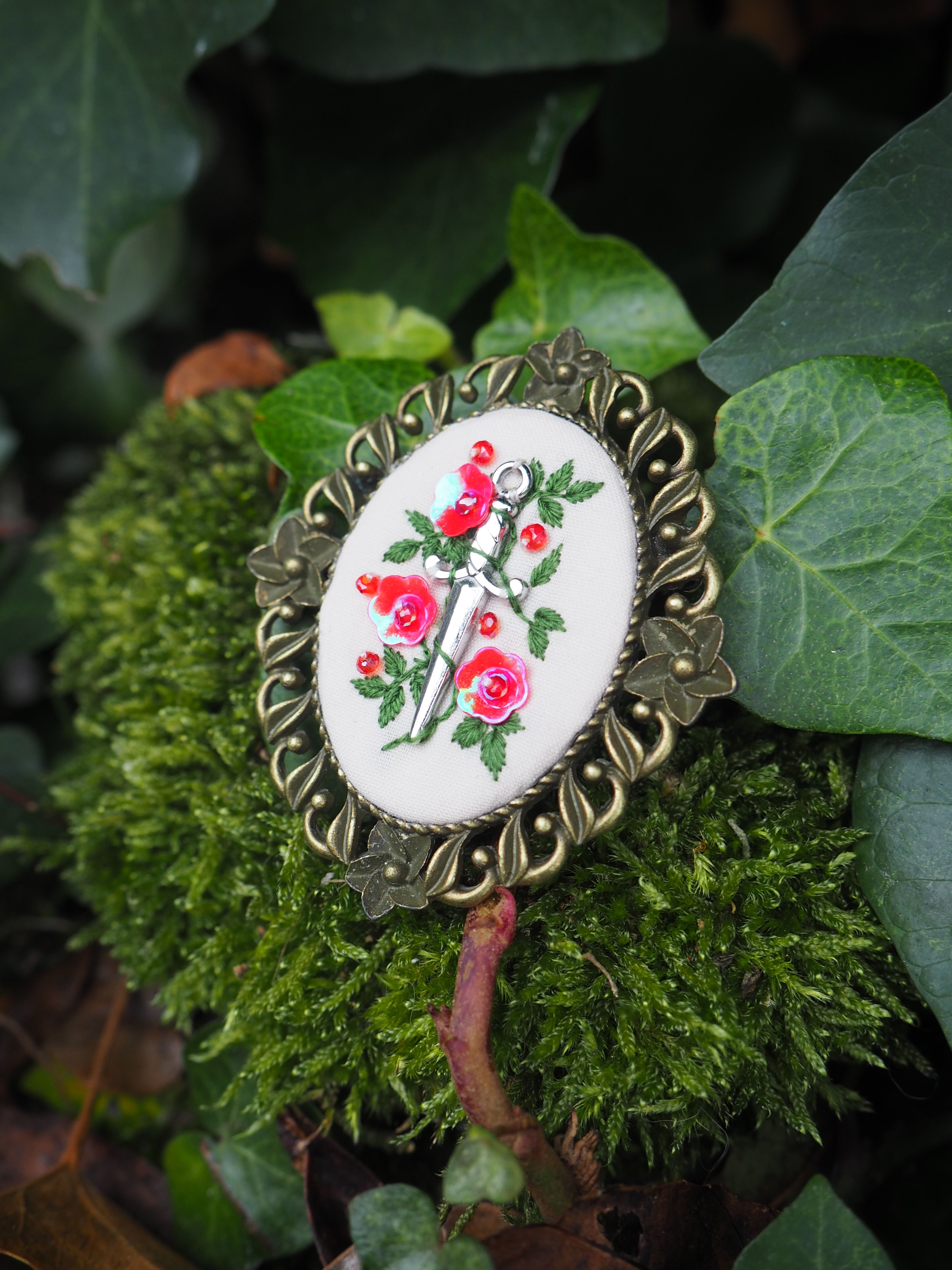 Cette image présente une broche décorative vintage posée sur un lit de mousse verte et entourée de feuillage naturel. La broche se compose d'un médaillon central ovale en porcelaine ou céramique blanche, orné d'un motif floral peint à la main représentant des roses rouges avec leurs feuilles vertes. Ce médaillon est serti dans un cadre métallique doré au style ornementé, avec des détails ciselés qui forment une bordure élégante autour du motif central. L'ensemble repose sur de la mousse d'un vert intense qui contraste joliment avec les couleurs de la broche, tandis que des feuilles vertes de différentes tailles et textures créent un arrière-plan naturel. L'éclairage doux met en valeur les détails délicats de la peinture florale et fait ressortir les reflets dorés du métal. Cette mise en scène évoque un style romantique et nostalgique, rappelant les bijoux d'époque ou les accessoires de mode rétro.