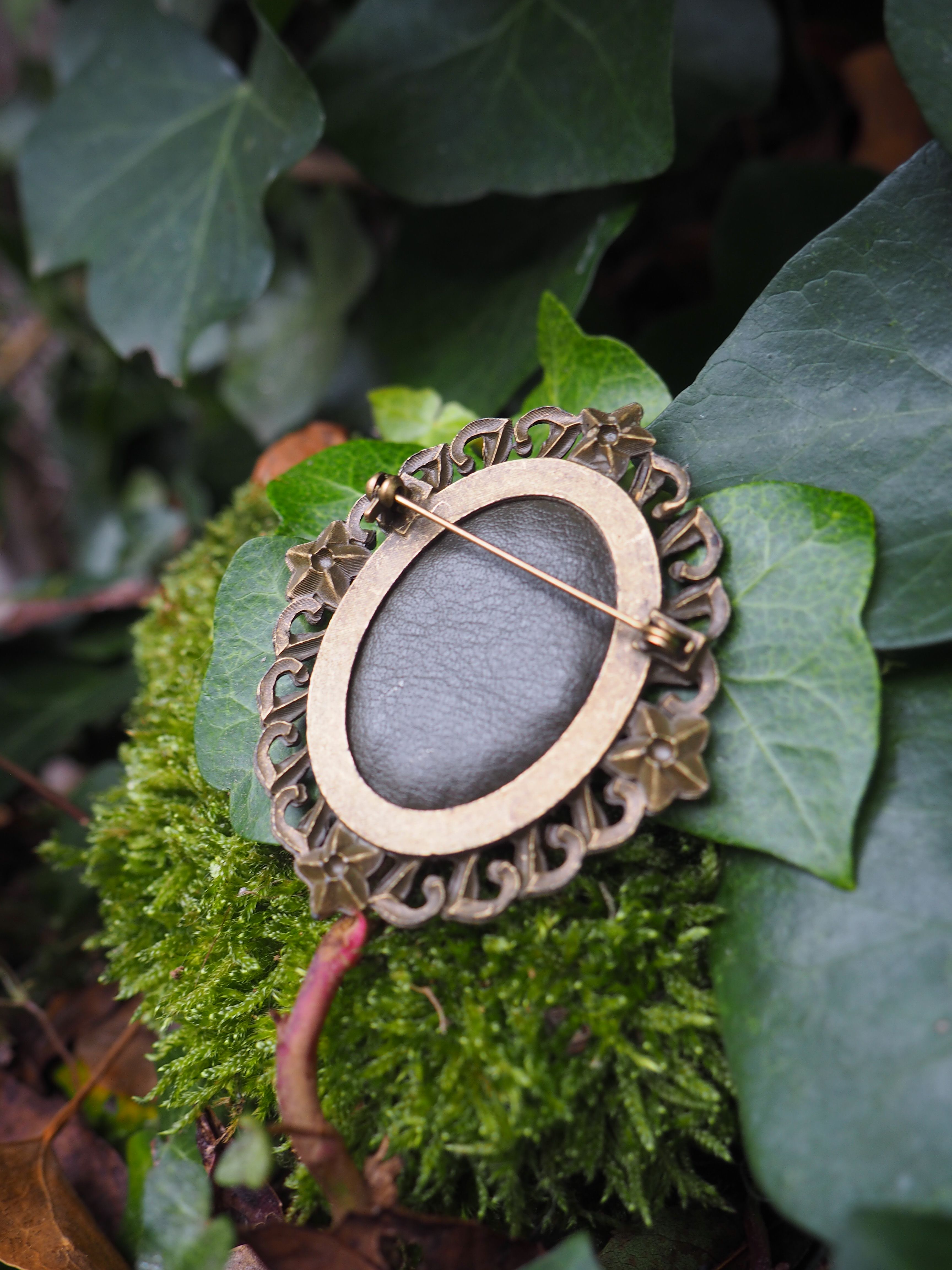 Cette image présente une broche ou un pendentif de style vintage posé délicatement sur un lit de mousse verte et de feuillage. Le bijou a une forme ovale avec un cadre décoratif en métal doré ou bronze, orné de motifs sculptés ou gravés qui lui donnent un aspect élégant et raffiné.  Au centre de la pièce se trouve une pierre ou un cabochon de couleur gris-violet, lisse et poli, qui contraste harmonieusement avec le métal doré du cadre. Une épingle métallique traverse le bijou, confirmant qu'il s'agit d'une broche fonctionnelle. L'ensemble repose sur un écrin naturel composé de mousse d'un vert tendre et de diverses feuilles aux nuances vertes variées, créant un arrière-plan organique qui met en valeur l'objet. Cette mise en scène évoque un style romantique ou bohème, où la nature sert d'écrin à ce bijou au charme rétro. L'éclairage doux et naturel de la photographie accentue les textures et les couleurs, créant une atmosphère poétique et intemporelle.