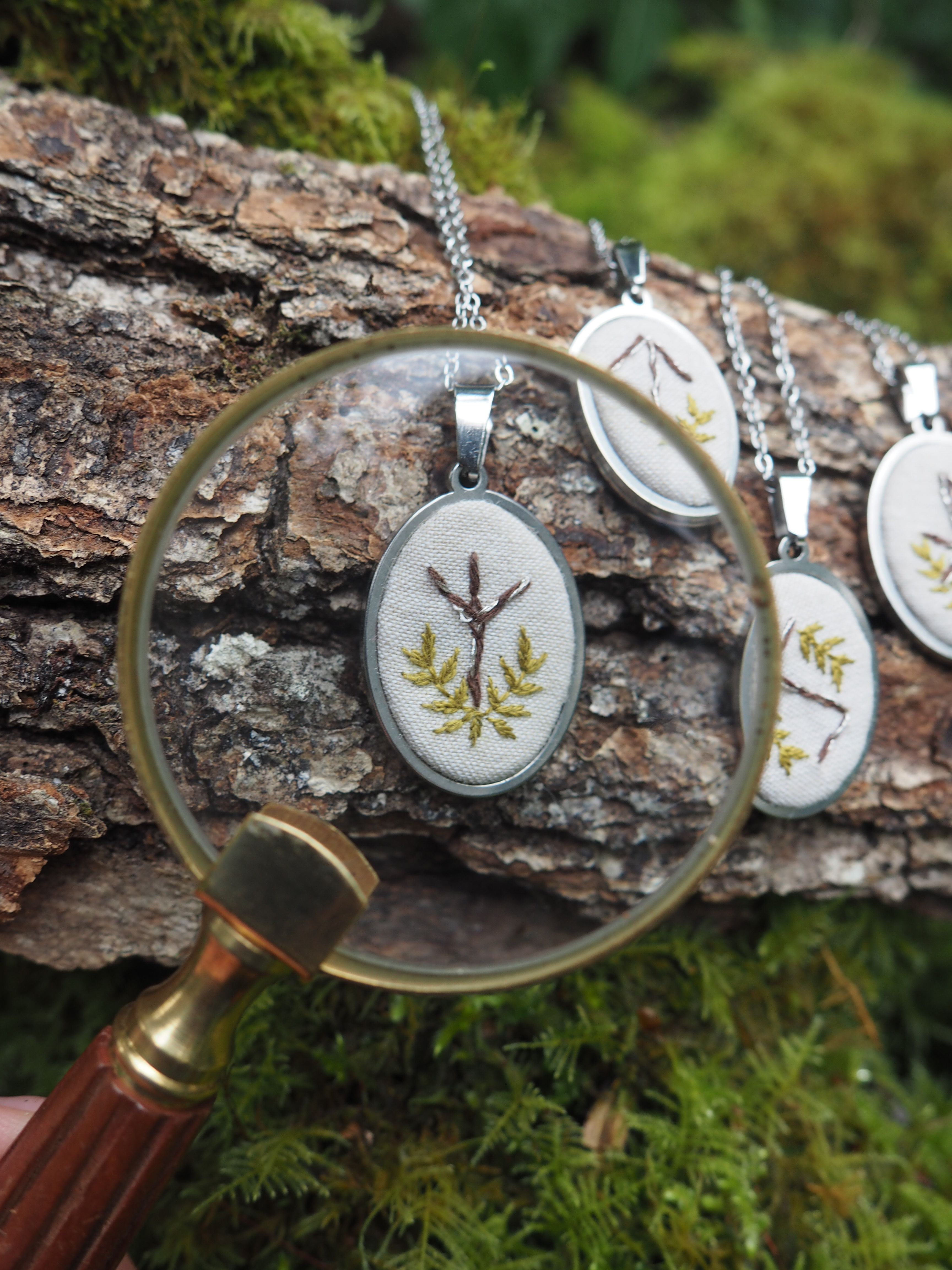 L'image présente une composition artistique et naturelle, centrée sur des pendentifs brodés, avec une loupe en premier plan qui attire l'attention sur l'un d'eux. Au premier plan, une loupe à monture dorée et manche en bois est tenue, son verre grossissant un pendentif ovale. Ce pendentif est brodé d'un motif végétal délicat : une branche stylisée avec des feuilles vertes et une forme qui ressemble à une petite branche ou une racine marron au centre, le tout sur un fond de tissu clair, probablement du lin ou du coton écru. La broderie est fine et détaillée. Derrière la loupe, plusieurs autres pendentifs similaires sont alignés sur un morceau d'écorce d'arbre rugueuse et texturée, qui sert de support naturel. Ces pendentifs sont également de forme ovale, avec des motifs brodés variés, mais tous dans le même style végétal et naturel. Ils sont suspendus à des chaînes fines, probablement en métal argenté. L'arrière-plan est flou, mais on distingue une végétation luxuriante et verdoyante, composée de mousse et de feuillage, ce qui renforce l'ambiance naturelle et forestière de la scène. La lumière semble douce et diffuse, mettant en valeur les textures et les couleurs des objets. L'ensemble de l'image évoque l'artisanat, la nature, la délicatesse et l'attention aux détails, suggérant peut-être une collection de bijoux faits à la main inspirés par le monde végétal.