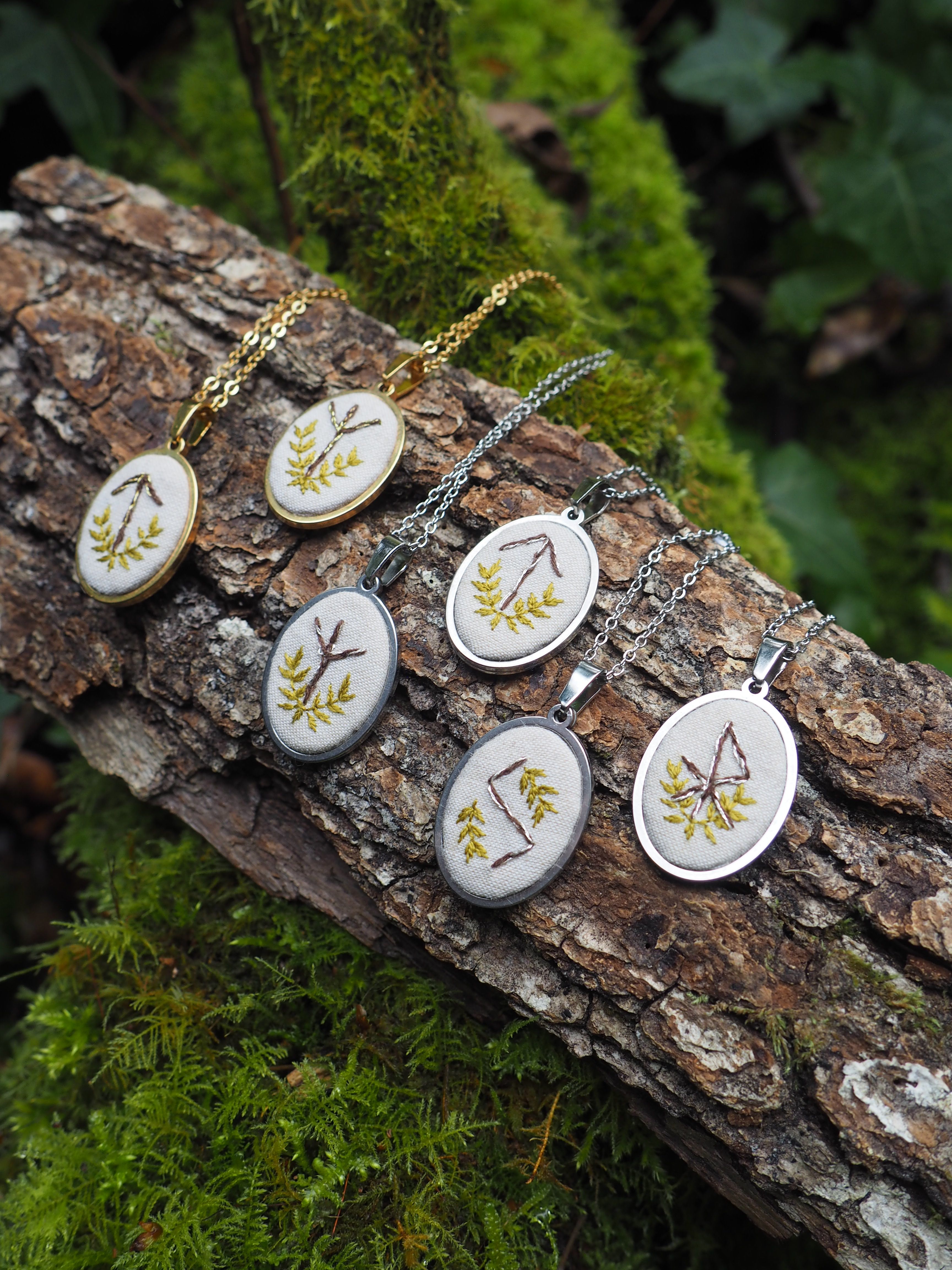 L'image présente six pendentifs ovales, chacun orné d'un motif brodé différent, posés sur un tronc d'arbre recouvert de mousse. Voici une description détaillée des éléments visuels : *   **Pendentifs :** Il y a six pendentifs de forme ovale. Chaque pendentif est composé d'un cadre métallique (certains dorés, d'autres argentés) et d'une base en tissu de couleur claire (blanc cassé ou crème) sur laquelle est brodé un motif.
    *   Les motifs brodés semblent être des symboles ou des lettres stylisées, réalisés avec du fil de couleur verte et marron, évoquant des branches, des feuilles ou des runes.
    *   Chaque pendentif est attaché à une chaîne fine, également métallique (dorée ou argentée), suggérant qu'il s'agit de colliers.
    *   Les pendentifs sont disposés en deux rangées légèrement décalées sur le tronc d'arbre. *   **Support (tronc d'arbre) :** Les pendentifs reposent sur un morceau de tronc d'arbre ou une grosse branche.
    *   L'écorce du bois est visible, avec des textures et des crevasses naturelles.
    *   Une grande partie du bois est recouverte de mousse verte, ajoutant une touche naturelle et organique à la scène. *   **Arrière-plan :** L'arrière-plan est flou, mais on distingue des éléments de végétation, principalement des feuilles vertes et de la mousse, ce qui renforce l'ambiance forestière ou naturelle de la photographie. La lumière semble douce et diffuse, typique d'un environnement boisé. *   **Couleurs et ambiance :** La palette de couleurs est dominée par les verts (mousse, broderies), les marrons (bois, broderies) et les tons métalliques (doré, argenté) des pendentifs et chaînes. L'ensemble dégage une atmosphère artisanale, naturelle et un peu mystique, potentiellement liée à des thèmes celtiques, nordiques ou de la nature. En résumé, l'image met en valeur une collection de pendentifs brodés à la main, présentés de manière rustique et naturelle sur un tronc d'arbre moussu, dans un cadre extérieur verdoyant.
