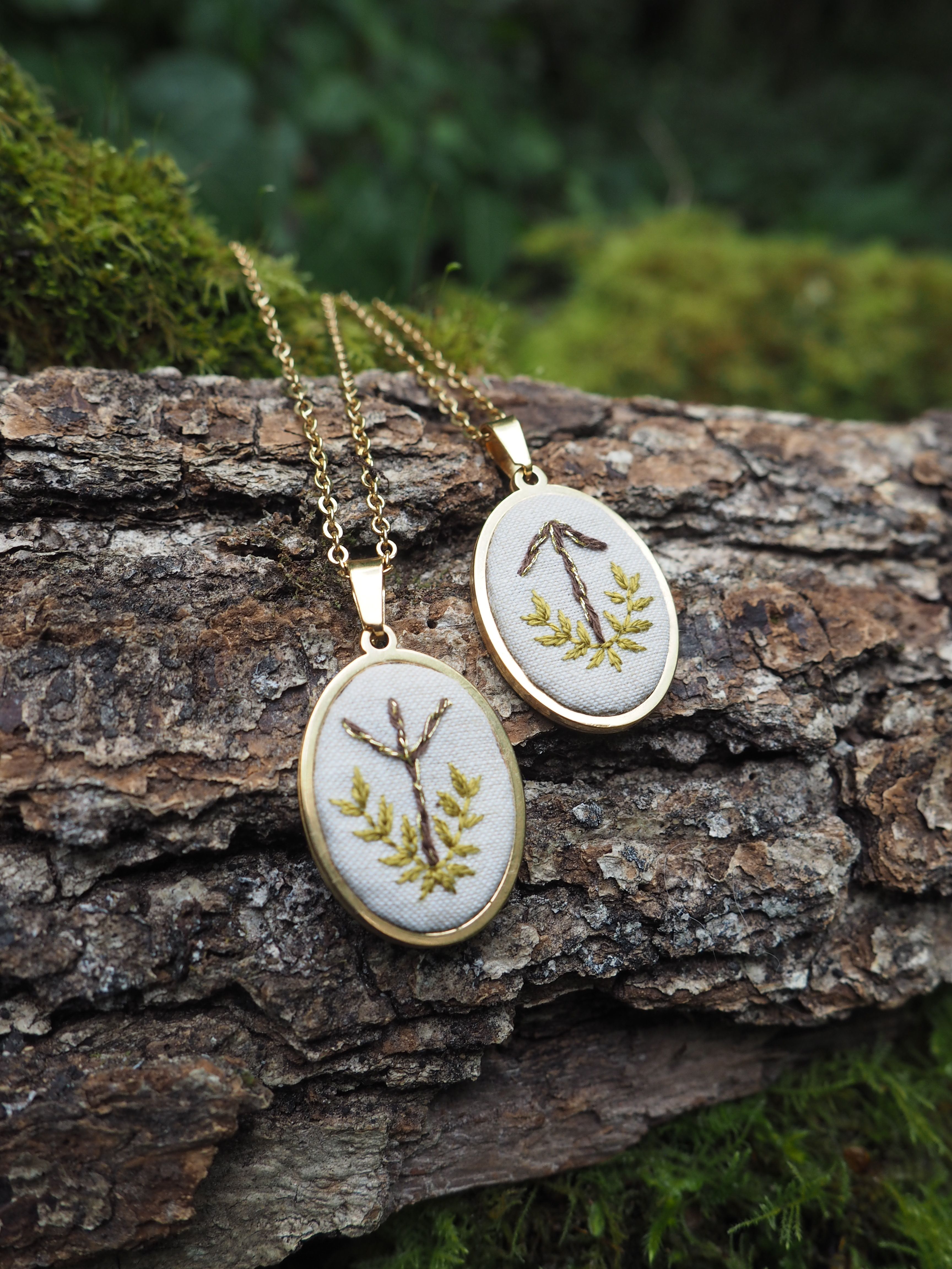 Cette image présente deux pendentifs ovales en laiton doré, chacun suspendu à une fine chaîne dorée. Les pendentifs sont posés sur un morceau de bois brut, probablement une bûche ou une branche, dont l'écorce est texturée et de couleur marron foncé. Chaque pendentif contient une broderie sur un fond de tissu clair, probablement blanc cassé ou crème. Les broderies représentent des motifs végétaux stylisés, évoquant des branches ou des feuilles.
Le pendentif de gauche montre un motif qui ressemble à une branche d'arbre avec plusieurs ramifications, brodée en fil marron et agrémentée de petites feuilles vertes ou dorées.
Le pendentif de droite présente un motif similaire, mais la forme principale est plus anguleuse, rappelant une ancre ou une lettre stylisée, également brodée en fil marron avec des feuilles vertes ou dorées de chaque côté de la base. L'arrière-plan est flou, mais on distingue des tons de vert et de marron, suggérant un environnement naturel, peut-être une forêt ou un jardin, avec de la mousse et de la végétation. La lumière semble naturelle et douce, mettant en valeur les détails des pendentifs. L'ensemble de l'image dégage une atmosphère artisanale et naturelle, soulignant la beauté des bijoux faits à la main et leur lien avec la nature.