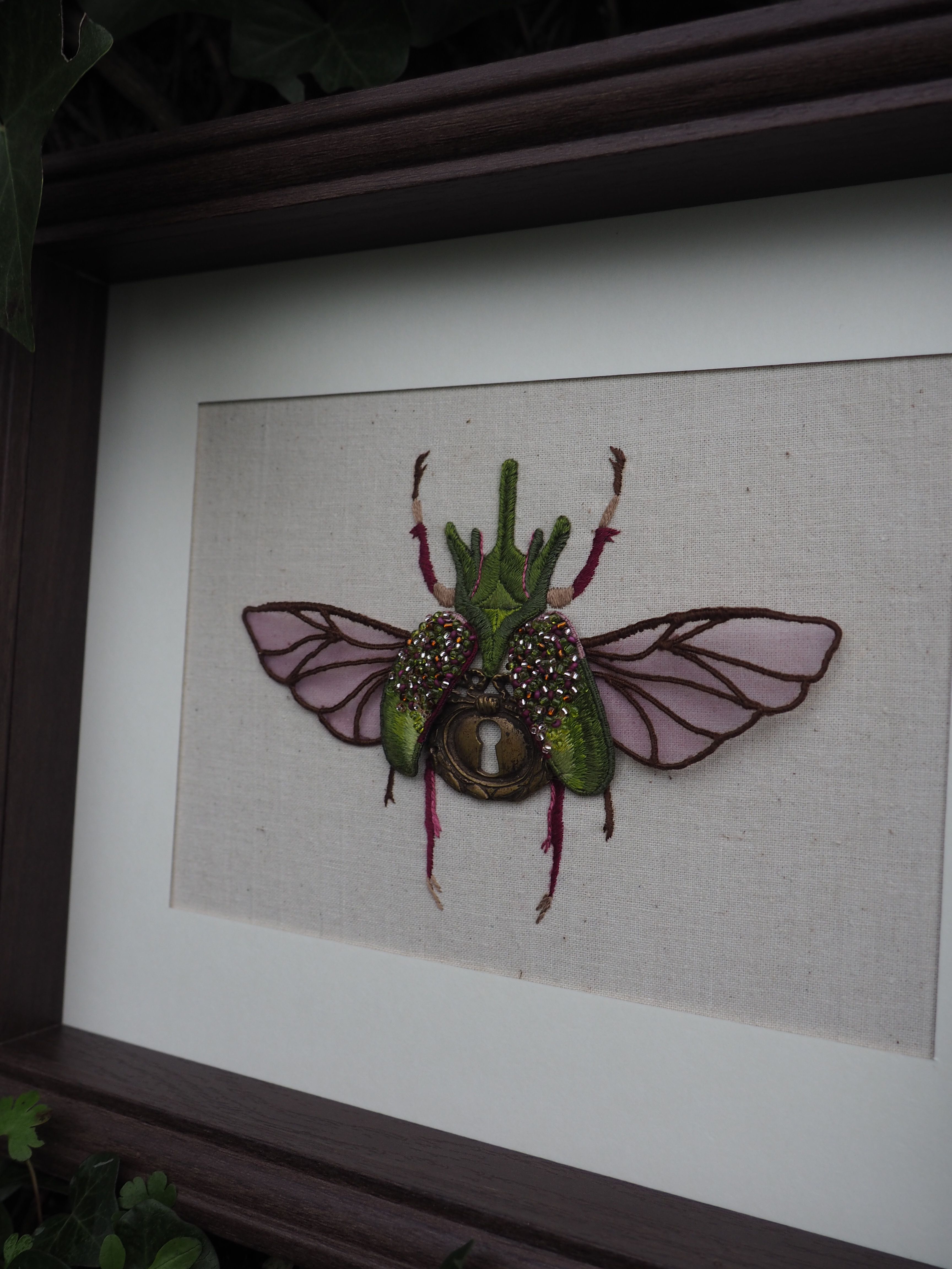 Cette image montre une œuvre d'art encadrée représentant un insecte décoratif. Il s'agit d'une création artistique d'un scarabée ou d'un coléoptère avec des ailes déployées, réalisée dans un style mixte combinant différents matériaux. L'insecte est de couleur verte avec des ailes translucides ou rosées, et semble avoir des détails brillants ou métalliques. Il est présenté sur un fond clair, probablement en tissu ou en papier, dans un cadre sombre. C'est une pièce décorative qui mélange l'aspect naturaliste des insectes avec une touche artistique et fantaisiste.