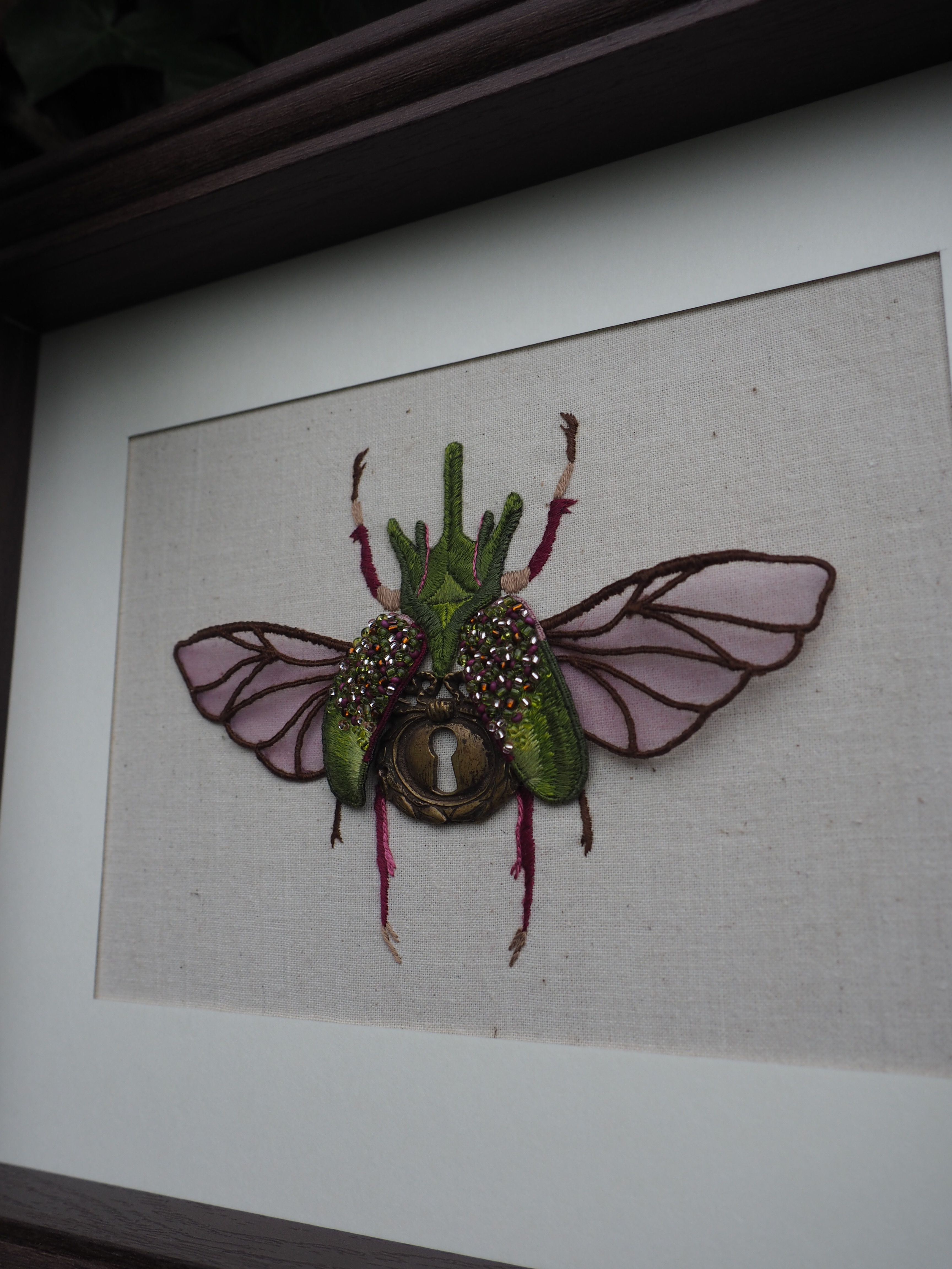 Cette image montre une œuvre d'art textile encadrée représentant un scarabée ou un insecte stylisé. L'insecte est créé avec différents matériaux et textures, notamment du tissu vert pour le corps, des ailes transparentes ou violettes, et ce qui semble être des éléments décoratifs brillants ou pailletés. Au centre de l'insecte se trouve ce qui ressemble à un œil ou une pierre décorative. L'œuvre est présentée sur un fond clair, probablement en lin ou en toile, et est encadrée dans ce qui semble être un cadre sombre. C'est une pièce décorative qui mélange l'art textile et l'entomologie artistique, créant un effet à la fois naturaliste et fantaisiste.