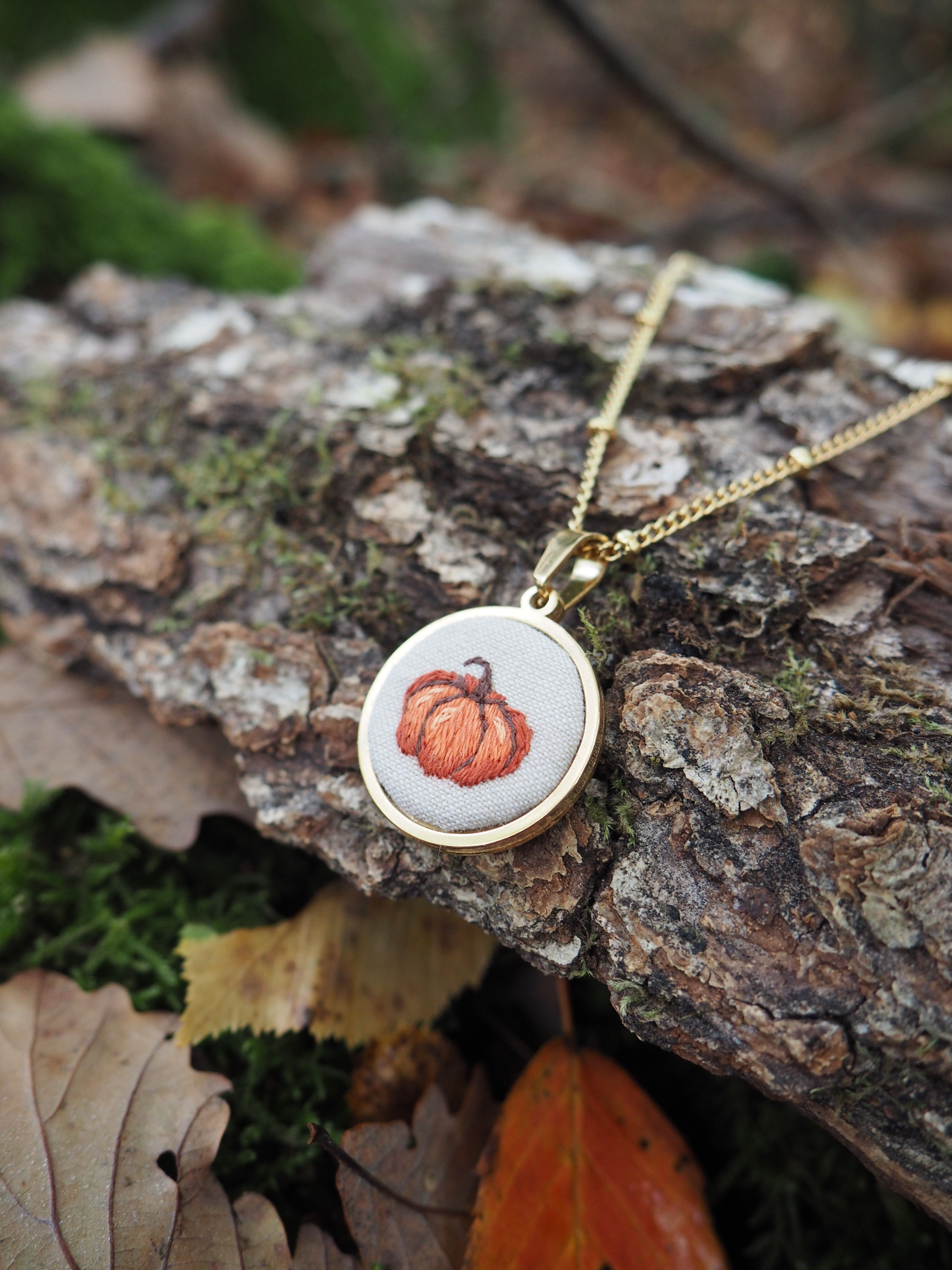 Cette image présente un collier délicat posé sur un morceau d'écorce d'arbre dans un environnement naturel automnal. Le pendentif circulaire, monté sur une chaîne dorée fine, arbore une illustration miniature d'une citrouille orange sur fond blanc ou crème.  L'écorce rugueuse et texturée sur laquelle repose le bijou contraste élégamment avec la finesse du pendentif. Autour, on aperçoit des feuilles mortes aux teintes automnales - brunes et orangées - qui s'harmonisent parfaitement avec le motif de citrouille du collier. L'arrière-plan flou révèle d'autres éléments naturels comme de la mousse verte et des feuilles, créant une composition qui évoque l'atmosphère chaleureuse de l'automne. Cette mise en scène naturelle met en valeur le caractère saisonnier du bijou, probablement conçu pour célébrer la saison automnale ou Halloween. La photographie capture bien les détails du pendentif tout en créant une ambiance rustique et naturelle qui complète parfaitement le thème automnal du bijou.