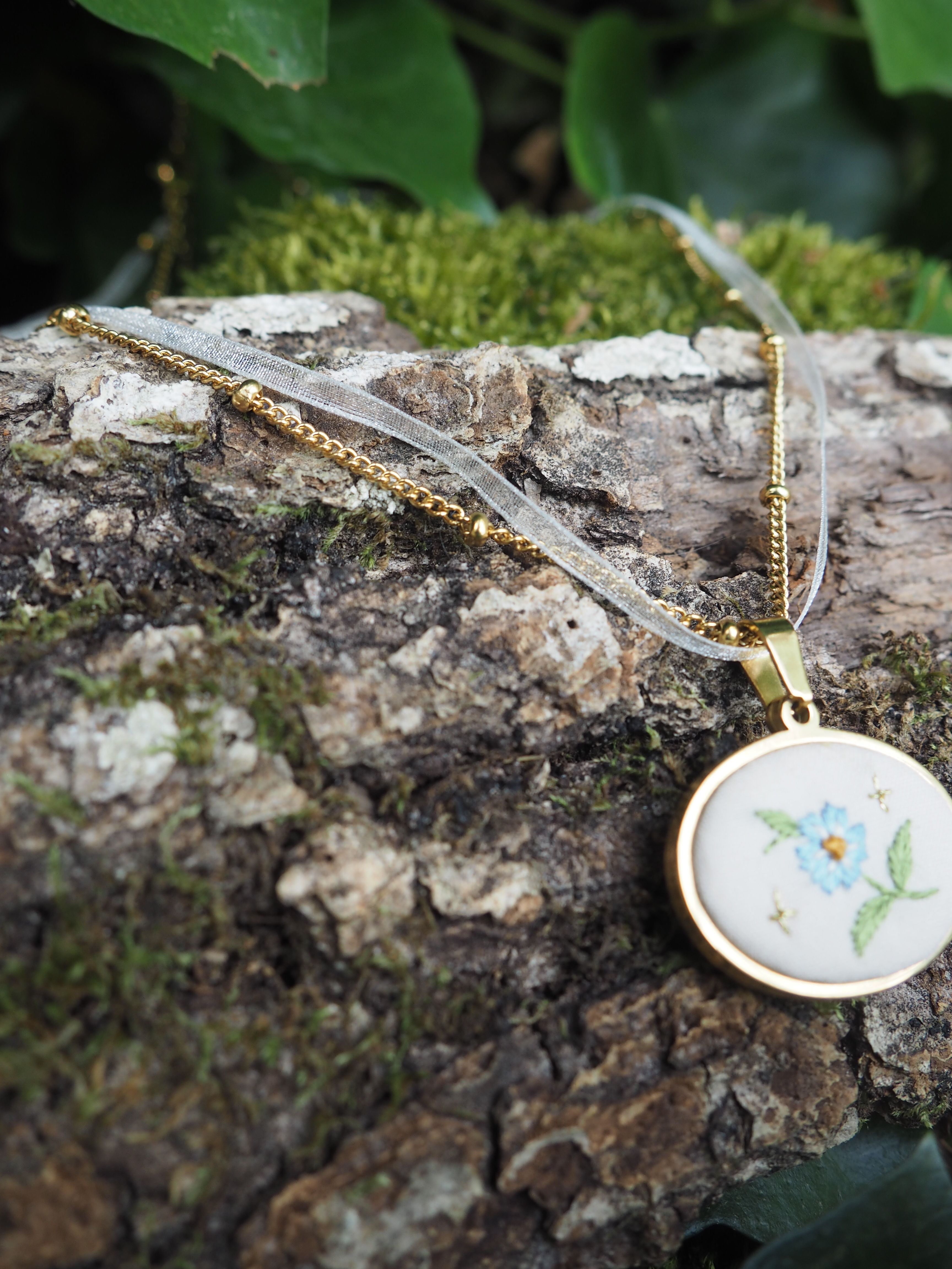 Cette image montre un délicat pendentif sur une chaîne dorée, posé sur un morceau d'écorce d'arbre couvert de mousse. Le pendentif est de forme ovale avec un fond blanc et comporte une délicate broderie florale, on peut distinguer une petite fleur bleue avec des feuilles vertes. La composition est très naturelle et poétique, avec le contraste entre le bijou raffiné et la texture rugueuse de l'écorce, ainsi que la verdure de la mousse en arrière-plan. C'est une mise en scène qui évoque un style romantique et bucolique.