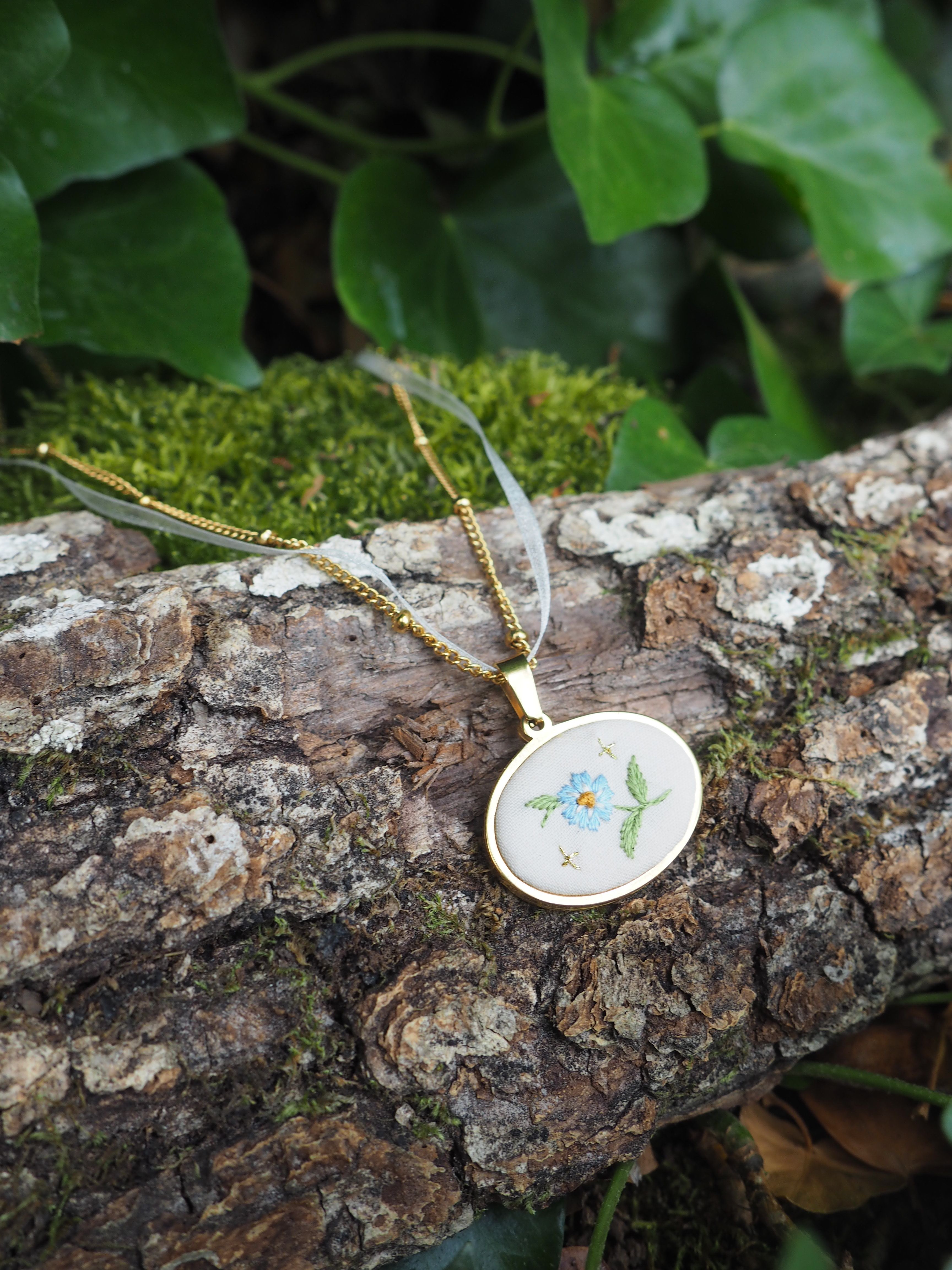 L'image montre des bijoux ou des pendentifs délicats posés sur une branche d'arbre couverte de mousse. Les pendentifs semblent être en porcelaine ou en céramique avec des motifs floraux peints en bleu clair. La branche a une écorce texturée et rugueuse, et on peut voir quelques feuilles vertes floues en arrière-plan. L'ensemble crée une composition naturelle et poétique, mélangeant des éléments naturels (le bois, la mousse) avec des pièces artisanales délicates.